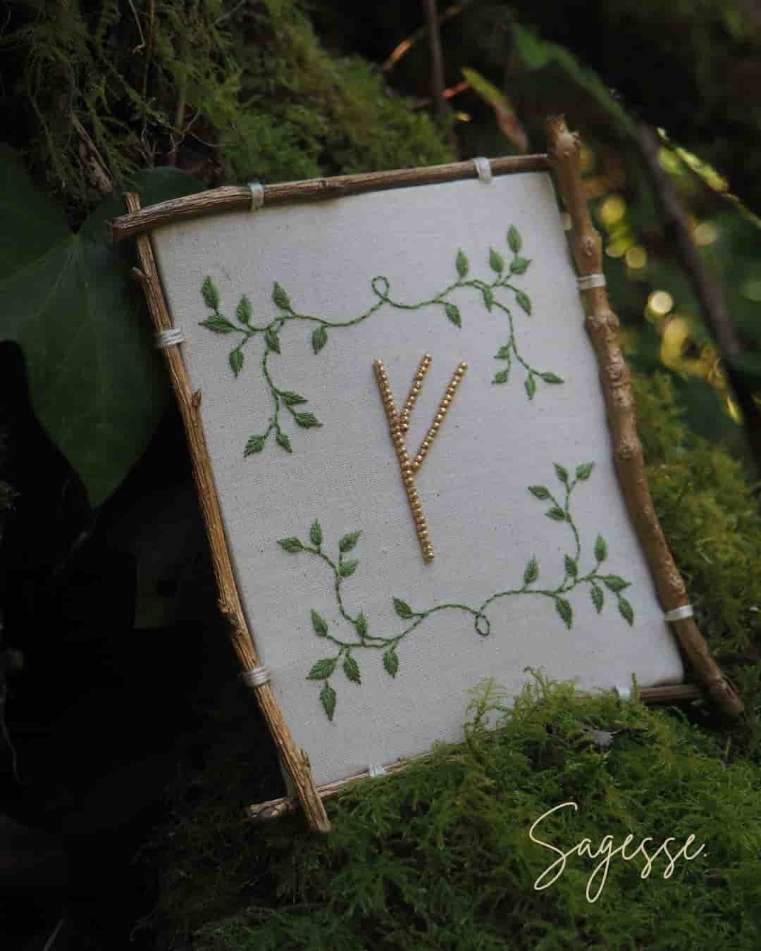 Cette image montre une belle composition artistique représentant la rune "Algiz" ou "Yr" (qui ressemble à un Y) brodée ou cousue sur un tissu blanc. Le symbole est entouré d'un motif délicat de feuilles et de vignes vertes qui s'entrelacent dans les coins. Le tissu est monté sur ce qui semble être un cadre rustique fait de petites branches ou de bois naturel. L'ensemble est photographié sur un fond de mousse verte et de feuillage, donnant une ambiance très naturelle et forestière. Le mot "Sagesse" apparaît en signature dans le coin inférieur droit de l'image. L'ensemble dégage une atmosphère très païenne, naturelle et mystique.