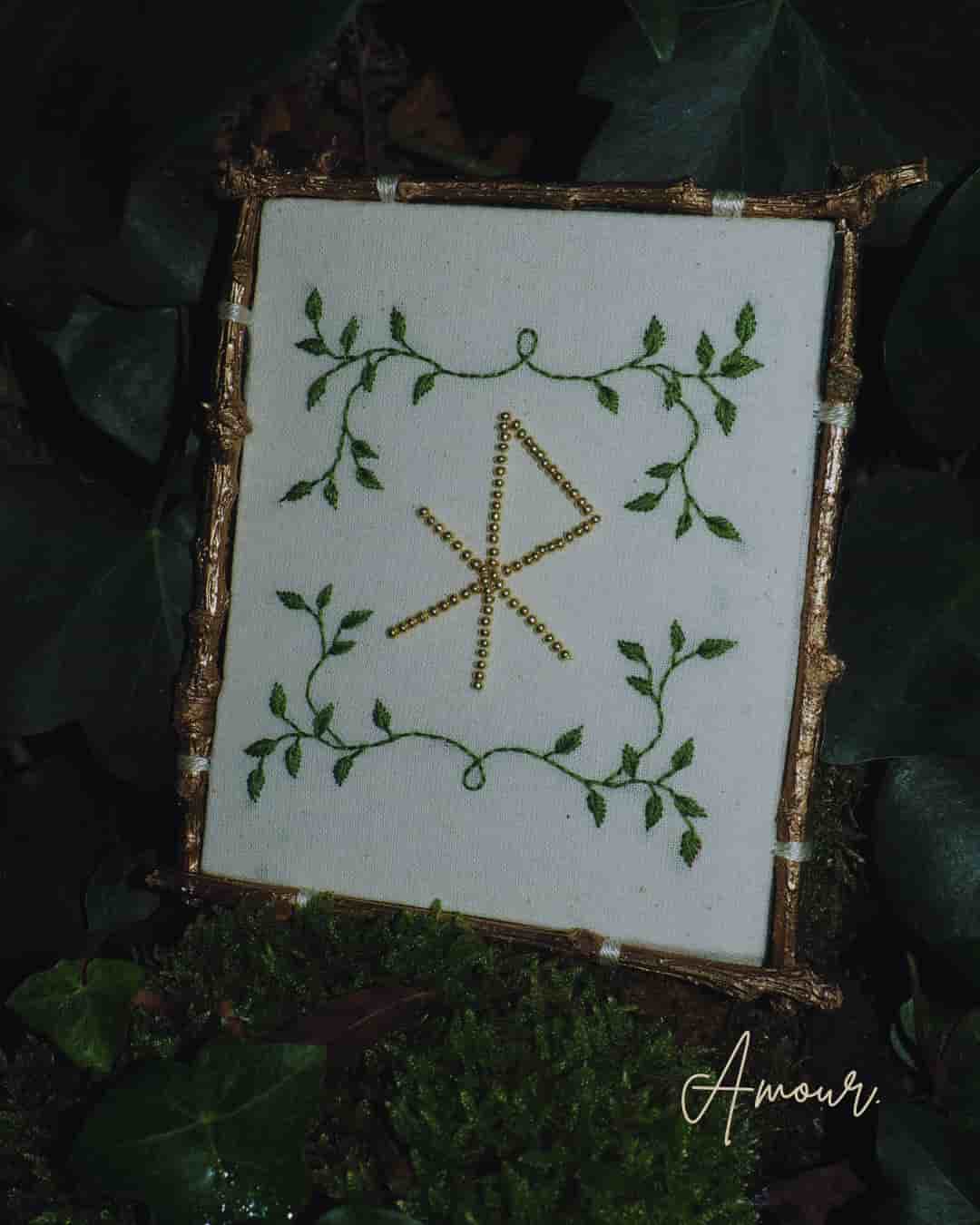 L'image présente une broderie encadrée, posée sur un lit de mousse et de feuilles de lierre, créant une ambiance naturelle et mystique. Au centre de l'image, un cadre de forme carrée ou légèrement rectangulaire, dont les bords semblent être faits de petites branches ou de brindilles dorées, contient une broderie sur un tissu de couleur crème ou blanc cassé. La broderie représente un motif central qui ressemble à une rune ou un symbole ancien, formé de lignes droites et obliques, réalisées avec des perles dorées ou des fils brillants. Autour de ce symbole central, des motifs de feuilles et de vrilles vertes sont brodés, encadrant le symbole et ajoutant une touche organique. Le cadre est posé sur un sol recouvert de mousse verte luxuriante et de feuilles de lierre vert foncé, qui s'étendent sur les côtés et en arrière-plan. La lumière semble douce et diffuse, créant des ombres subtiles et mettant en valeur les textures des éléments naturels. Dans le coin inférieur droit de l'image, le mot "Amour" est écrit en cursive dorée ou jaune pâle, ajoutant une dimension émotionnelle ou thématique à la scène. L'ensemble de l'image dégage une atmosphère de calme, de nature et de spiritualité, avec une touche d'élégance rustique.