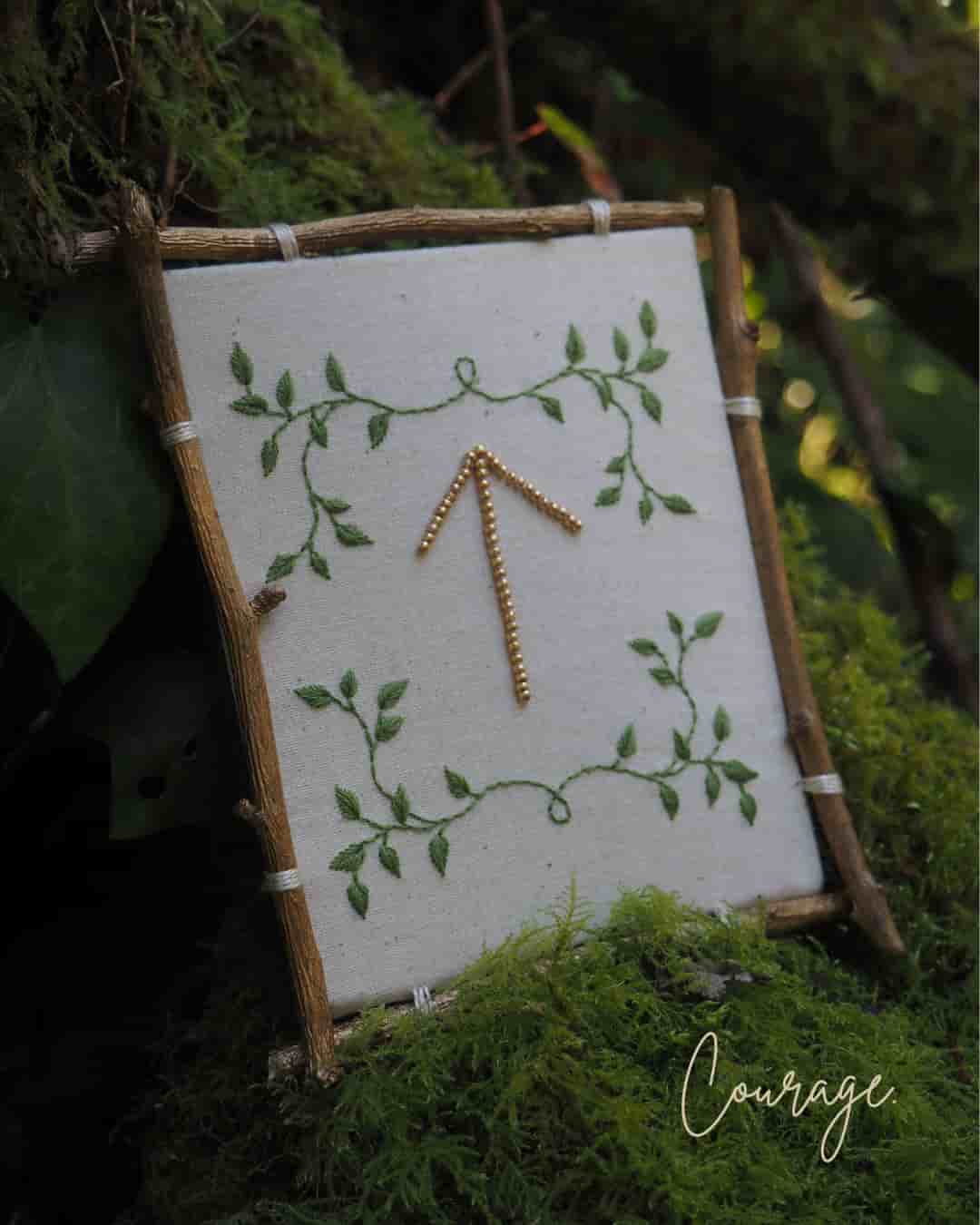 Cette image montre une création artisanale délicate : un symbole de rune ou une flèche pointant vers le haut, réalisée avec ce qui semble être des petites perles ou des bâtonnets dorés. Ce motif est encadré par des branches vertes brodées ou peintes sur un fond blanc. Le tout est présenté dans un cadre rustique fait de petites branches naturelles assemblées. L'œuvre est photographiée sur un lit de mousse verte, donnant une ambiance très naturelle et forestière à l'ensemble. Le mot "Courage" apparaît en écriture cursive dans le coin inférieur droit de l'image. L'ensemble dégage une atmosphère paisible et inspirante, mêlant éléments naturels et artisanat délicat.