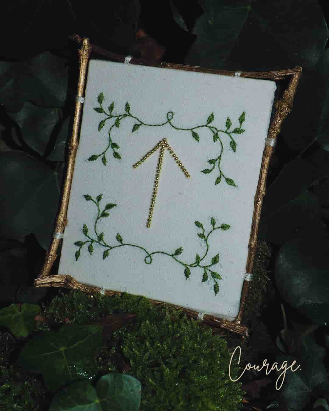 L'image présente une broderie encadrée, posée sur un lit de mousse et de feuilles de lierre, créant une ambiance naturelle et forestière. Au centre de l'image, un cadre rustique, probablement fait de brindilles ou de petits morceaux de bois dorés, encadre une pièce de tissu de couleur crème ou blanc cassé. Sur ce tissu, une broderie délicate est visible. Le motif principal est une flèche pointant vers le haut, brodée avec des perles ou des fils dorés, lui donnant un aspect scintillant. Autour de cette flèche, des motifs de feuilles vertes sont brodés, formant des guirlandes ou des arabesques qui encadrent la flèche, renforçant le thème végétal. Le cadre est posé sur un tapis de mousse verte luxuriante, parsemé de feuilles de lierre vert foncé, certaines avec des reflets brillants. L'arrière-plan est flou, mais on distingue des feuillages sombres, suggérant un environnement boisé ou un jardin. Dans le coin inférieur droit de l'image, le mot "Courage" est écrit en lettres cursives dorées ou de couleur claire, ajoutant une signification ou un message à la scène. L'éclairage semble doux et naturel, mettant en valeur les textures de la mousse, des feuilles et la brillance de la broderie dorée. L'ensemble de l'image dégage une atmosphère de calme, de nature et de symbolisme.