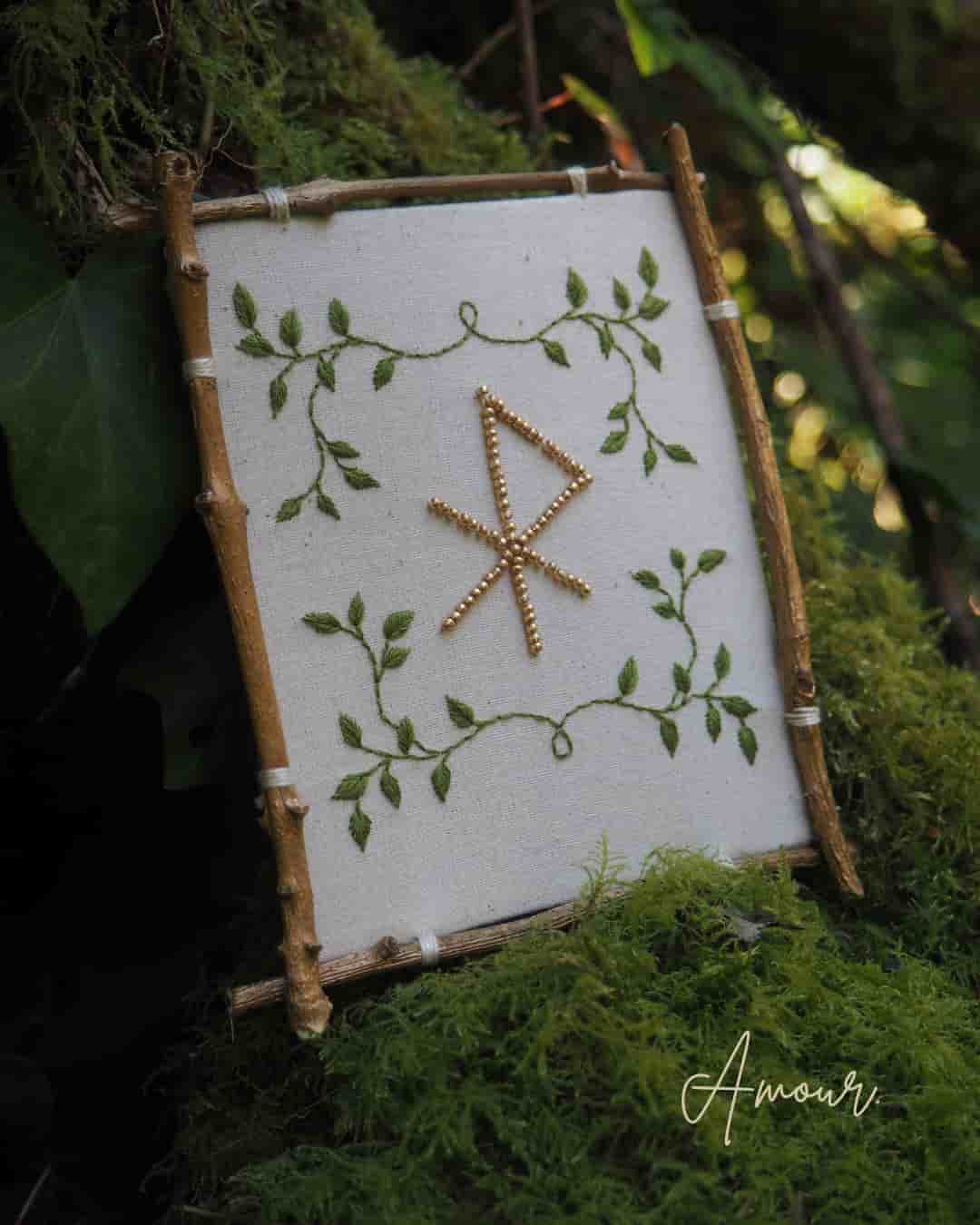 Cette image montre un ouvrage de broderie encadré dans un cadre rustique fait de petites branches ou bâtons de bois. La broderie est réalisée sur un tissu blanc et présente un motif central qui ressemble à une rune ou un symbole nordique fait de points dorés ou de fils métalliques. Ce symbole est entouré d'une délicate broderie de feuilles et de tiges vertes qui forment un motif végétal décoratif aux quatre coins. L'ensemble est photographié sur un fond naturel de mousse et de verdure, créant une ambiance féerique et forestière. Le mot "Amour" apparaît en signature dans le coin inférieur droit de l'image.