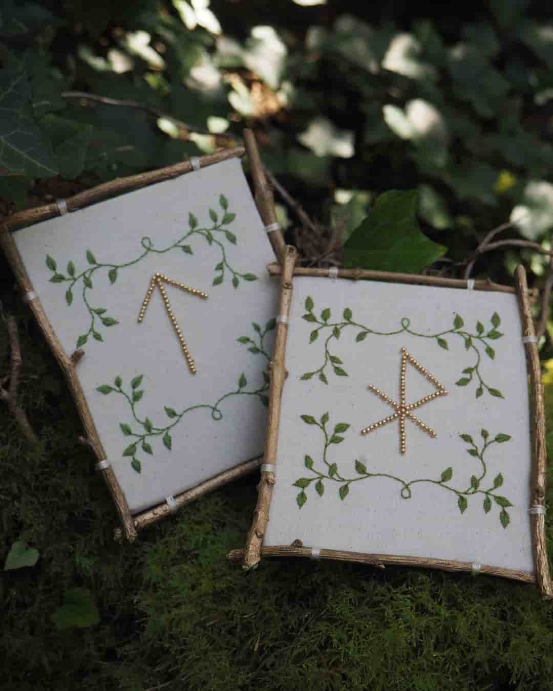 L'image présente deux cadres carrés, chacun contenant une broderie sur un tissu de couleur claire, probablement du lin ou du coton. Les cadres sont faits de petites branches ou de brindilles, ce qui leur donne un aspect rustique et naturel. Sur le cadre de gauche, la broderie représente une rune, qui ressemble à une flèche pointant vers le haut, faite de perles dorées. Autour de cette rune, il y a un motif de feuilles vertes brodées, formant une sorte de vigne ou de guirlande. Sur le cadre de droite, une autre rune est brodée avec des perles dorées. Cette rune a une forme plus complexe, avec des lignes diagonales et horizontales. Elle est également entourée d'un motif de feuilles vertes brodées, similaire à celui du cadre de gauche. Les deux cadres sont posés sur une surface recouverte de mousse verte, ce qui renforce l'ambiance naturelle et forestière de la scène. En arrière-plan, on peut distinguer des feuilles de lierre et d'autres végétaux, suggérant un environnement extérieur ou un décor inspiré de la nature. L'éclairage semble doux et naturel, mettant en valeur les détails des broderies et la texture des cadres en bois. L'ensemble de l'image dégage une atmosphère calme, artisanale et un peu mystique, évoquant peut-être des thèmes liés à la nature, à la spiritualité ou à l'artisanat traditionnel.