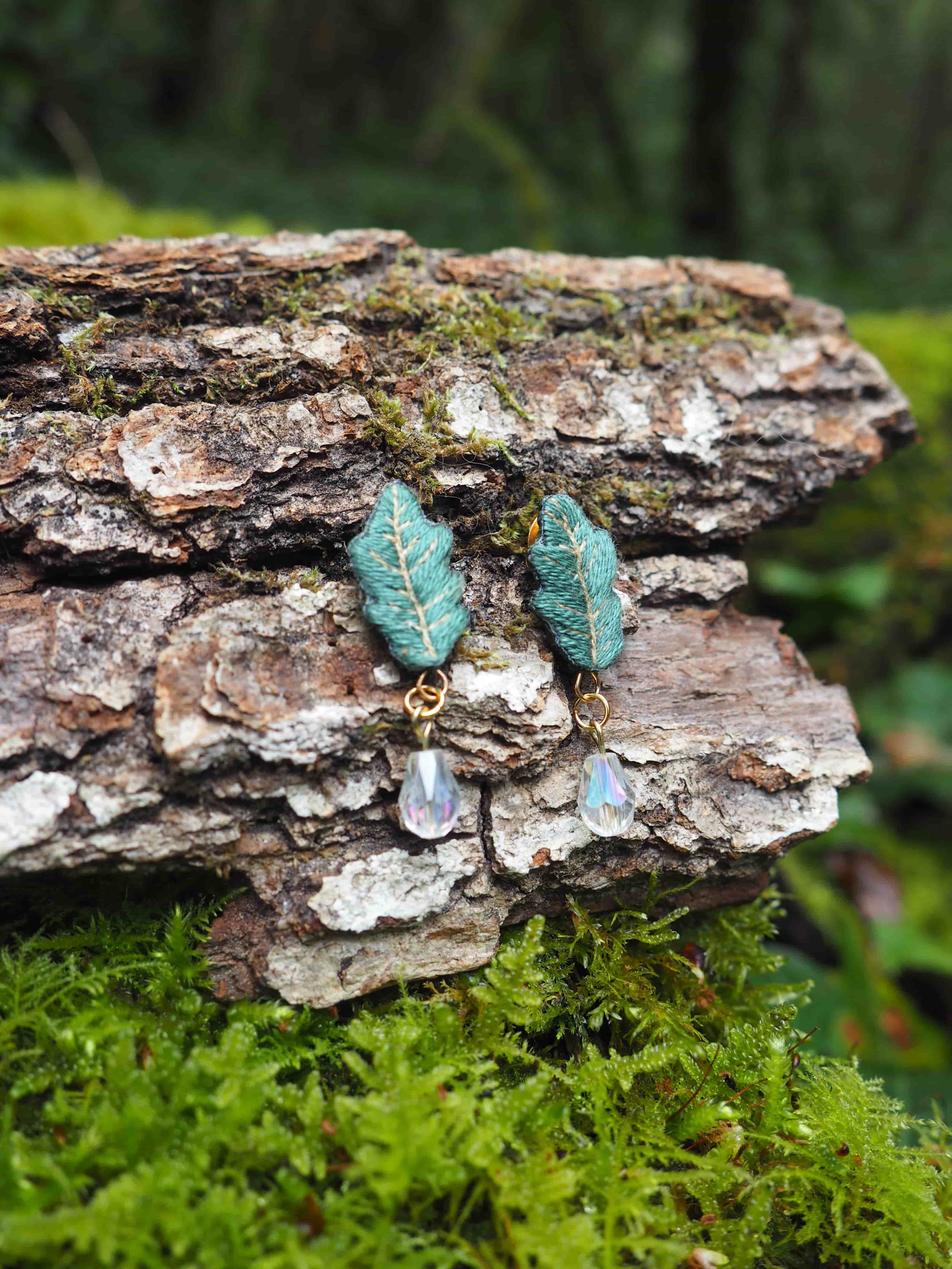 L'image présente une paire de boucles d'oreilles pendantes, posées sur un morceau d'écorce d'arbre texturée, recouverte de mousse verte. Chaque boucle d'oreille est composée d'un motif en forme de feuille de chêne stylisée, de couleur vert-bleu ou sarcelle, avec des détails de nervures apparents, suggérant une texture brodée ou sculptée. Sous chaque feuille pend une petite perle transparente et facettée, qui semble capter la lumière, ajoutant une touche d'éclat. Les attaches des boucles d'oreilles sont de couleur dorée. L'arrière-plan est flou, mais on distingue des tons de vert et de brun, indiquant un environnement naturel, probablement une forêt ou un sous-bois, ce qui complète le thème naturel des boucles d'oreilles. La lumière semble douce et diffuse, mettant en valeur les détails des bijoux et la texture de l'écorce et de la mousse.