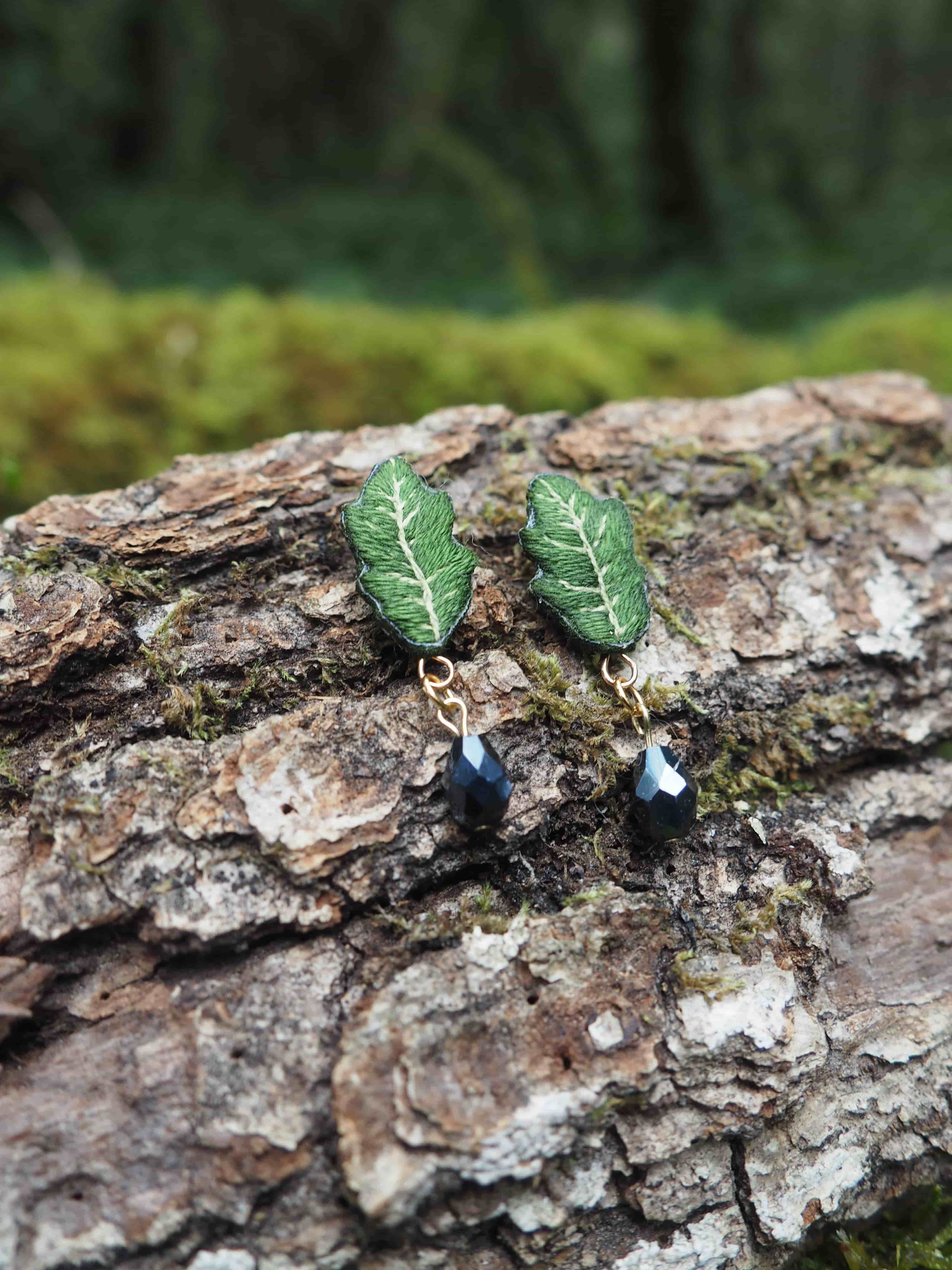 L'image présente une paire de boucles d'oreilles artisanales posées sur un morceau d'écorce d'arbre. **Description des boucles d'oreilles :**
*   Chaque boucle d'oreille est composée d'un motif supérieur en forme de feuille verte. Ces feuilles ont une texture détaillée, avec des nervures blanches ou très claires qui contrastent avec le vert foncé de la feuille, leur donnant un aspect réaliste et texturé.
*   Sous chaque feuille, pend une petite perle ou un cristal de couleur sombre, probablement noir ou gris foncé, qui capte la lumière et ajoute une touche d'éclat.
*   Les éléments sont reliés par de petits anneaux ou tiges métalliques de couleur dorée, qui ajoutent une touche d'élégance. **Contexte et arrière-plan :**
*   Les boucles d'oreilles sont posées sur un morceau d'écorce d'arbre rugueuse et texturée, de couleur marron clair à grisâtre, avec des crevasses et des aspérités. On peut apercevoir de petites touches de mousse verte sur l'écorce, renforçant le thème naturel.
*   L'arrière-plan est flou, mais on distingue des teintes de vert et de marron, suggérant un environnement forestier ou végétal, ce qui complète parfaitement le design des boucles d'oreilles en forme de feuille. **Ambiance générale :**
L'image dégage une ambiance naturelle, artisanale et délicate. La mise au point est nette sur les boucles d'oreilles, les mettant en valeur comme des objets uniques et faits à la main, inspirés par la nature.