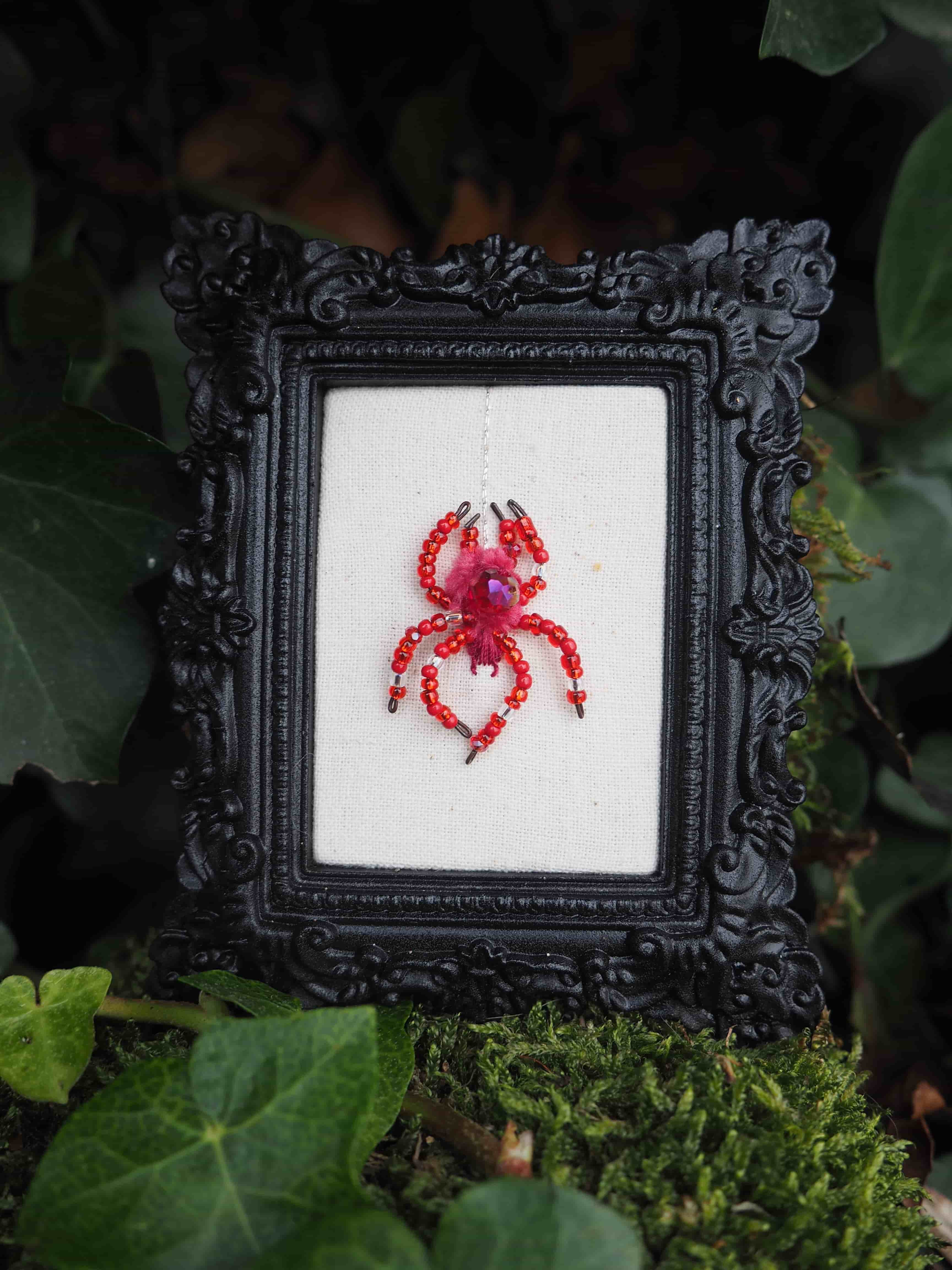 Cette image présente une œuvre d'art miniature mise en scène dans un environnement naturel. Au centre, on découvre un petit cadre noir orné de motifs décoratifs baroques qui encadre une broderie ou un point de croix représentant un crabe rouge et rose sur un fond de tissu blanc ou écru. Le cadre est artistiquement disposé sur un lit de mousse verte, entouré de feuillage naturel qui crée un contraste saisissant entre l'œuvre artisanale délicate et l'environnement organique. Les feuilles vertes qui l'entourent ajoutent une dimension naturelle et poétique à la composition. L'éclairage doux met en valeur les détails fins de la broderie du crustacé, dont les nuances de rouge et de rose ressortent particulièrement bien contre le fond clair. Cette mise en scène évoque une découverte précieuse cachée dans la nature, créant un dialogue intéressant entre l'art décoratif et l'environnement forestier. L'ensemble dégage une atmosphère féerique et mystérieuse, comme si l'on avait découvert un trésor artistique niché secrètement dans un coin de forêt.