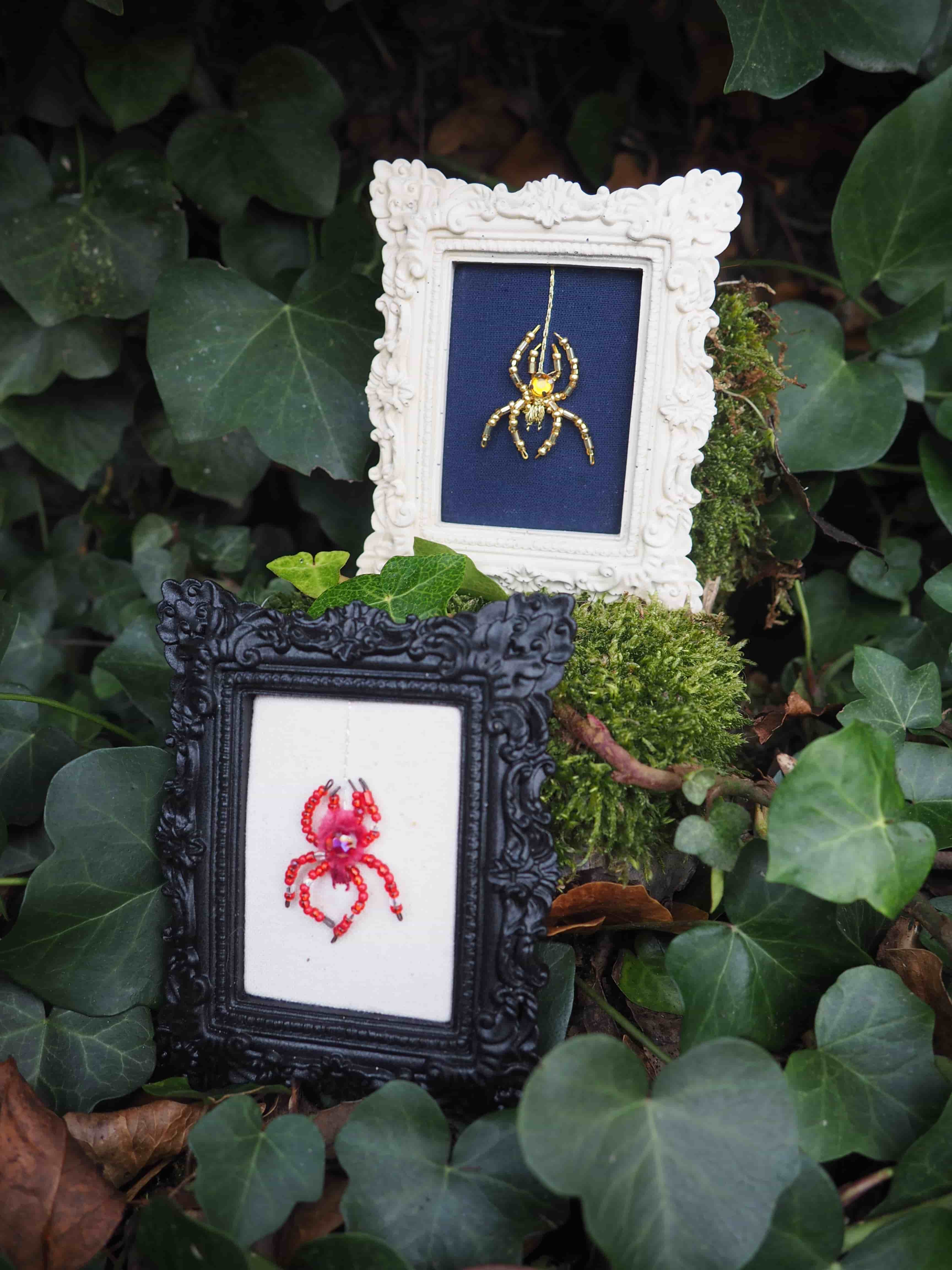 Cette image présente deux petits tableaux brodés disposés dans un environnement naturel luxuriant.  **Les œuvres :**
- **Cadre blanc orné** : Contient une broderie représentant une araignée dorée sur fond bleu marine. Le cadre présente des détails décoratifs en relief qui lui donnent un aspect vintage et élégant.
- **Cadre noir sculpté** : Abrite une broderie d'un crabe rouge sur fond blanc. Le cadre noir arbore des motifs ornementaux complexes qui créent un bel effet de contraste. **L'environnement :**
Les deux œuvres sont artistiquement disposées au milieu d'une végétation dense composée de :
- Grandes feuilles vertes (qui semblent être du lierre ou une plante similaire)
- Mousse verte qui tapisse le sol
- Feuilles mortes qui ajoutent des tons chauds à la composition **L'effet artistique :**
Cette mise en scène crée un contraste saisissant entre l'art décoratif raffiné des broderies et l'environnement naturel sauvage, évoquant peut-être le thème des créatures dans leur habitat naturel. La composition photographique met en valeur à la fois le travail artisanal minutieux et la beauté de la nature environnante.