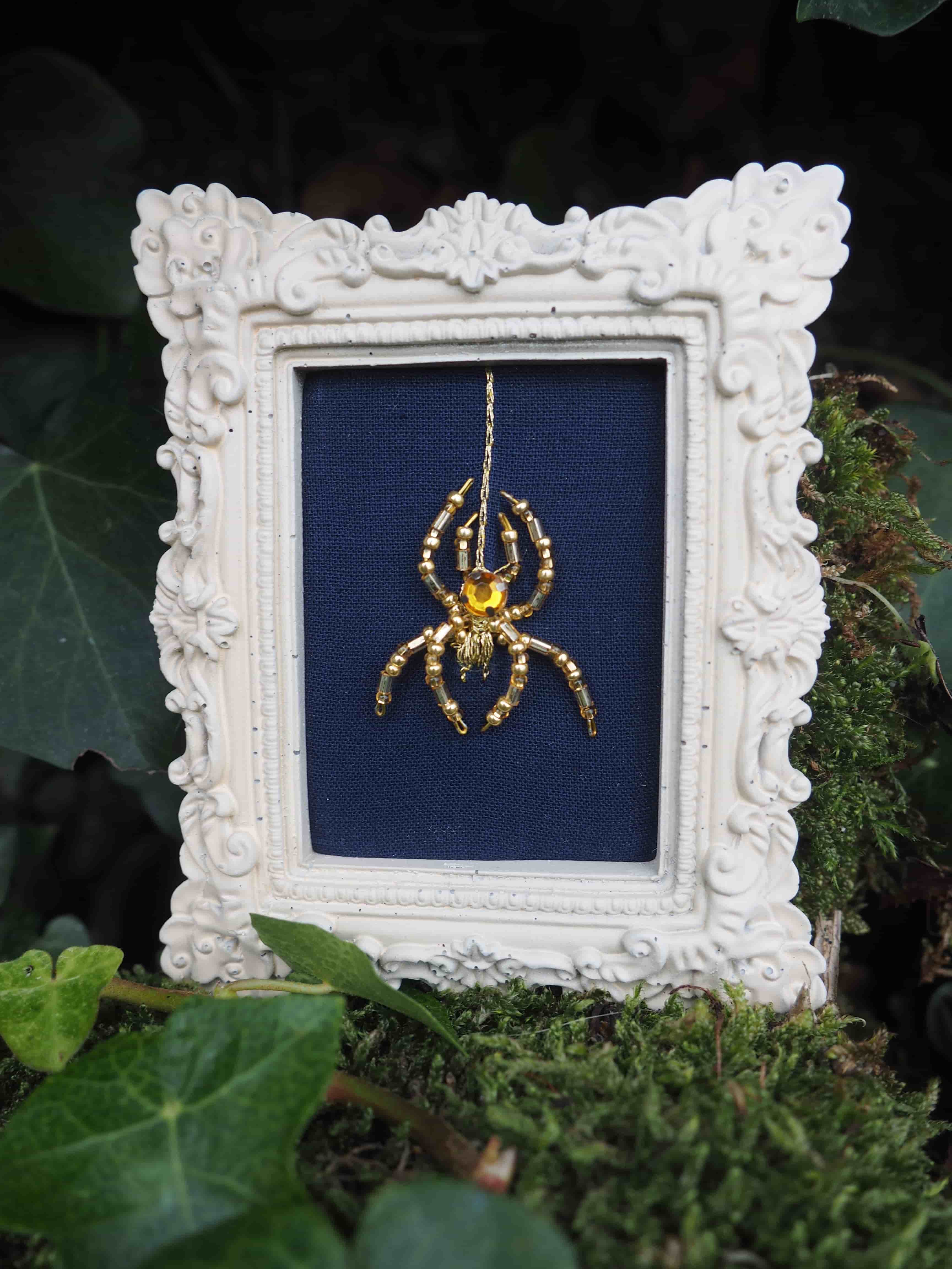 Cette image présente une œuvre d'art décorative encadrée, photographiée dans un environnement naturel verdoyant.  **Le cadre** : Il s'agit d'un cadre baroque blanc orné de motifs sculptés élaborés, avec des volutes, des fleurs et des détails décoratifs en relief qui lui donnent un aspect très ornemental et classique. **L'œuvre** : À l'intérieur du cadre se trouve une broderie ou un travail textile sur fond bleu marine. Le motif central représente une araignée dorée, probablement brodée au fil d'or ou doré, qui se détache élégamment sur le tissu sombre. **L'environnement** : L'ensemble est photographié posé sur de la mousse verte, entouré de feuillage et de végétation, créant un contraste saisissant entre l'objet d'art raffiné et l'environnement naturel sauvage. Cette mise en scène crée une atmosphère mystérieuse et poétique, mêlant l'artisanat délicat à la nature, avec l'araignée dorée comme symbole central qui fait écho à l'environnement naturel tout en étant sublimée par l'art textile.