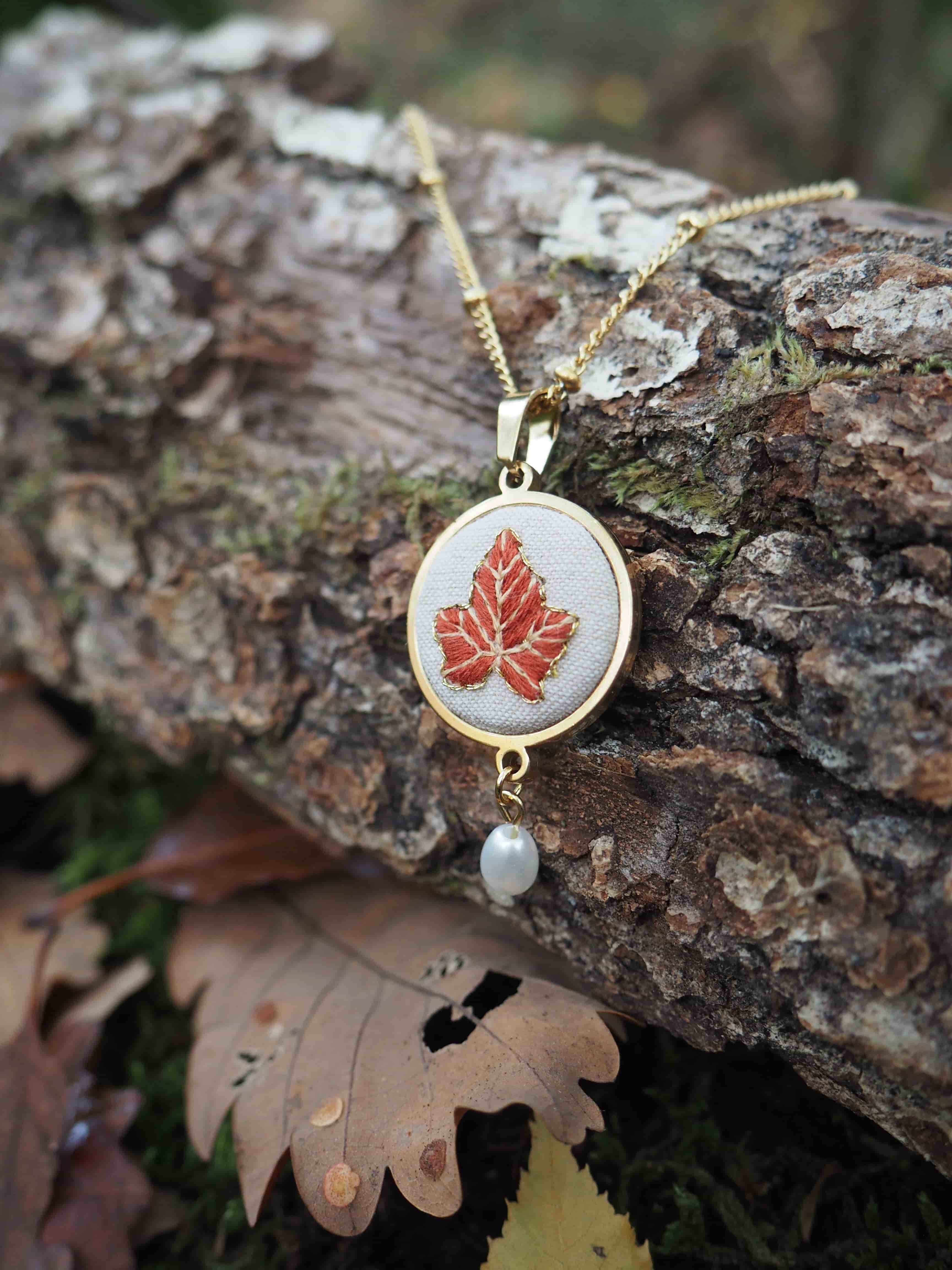 Cette image présente un élégant collier posé sur une bûche de bois dans un environnement naturel automnal. Le bijou se compose d'un pendentif circulaire avec un cadre doré qui encadre un motif de feuille d'érable rouge brodée ou imprimée sur un fond blanc ou crème. Une petite perle blanche pend délicatement sous le pendentif principal. Le collier repose sur une chaîne dorée fine qui serpente sur l'écorce rugueuse et texturée du bois. L'arrière-plan flou révèle des feuilles d'automne tombées au sol, créant une harmonie parfaite entre le motif de la feuille d'érable du bijou et l'environnement naturel qui l'entoure. La photographie capture magnifiquement les tons chauds de l'automne - les dorés du métal, les rouges de la feuille d'érable, et les bruns naturels du bois et des feuilles mortes. Cette mise en scène évoque la beauté saisonnière et donne au bijou une dimension poétique et naturelle.