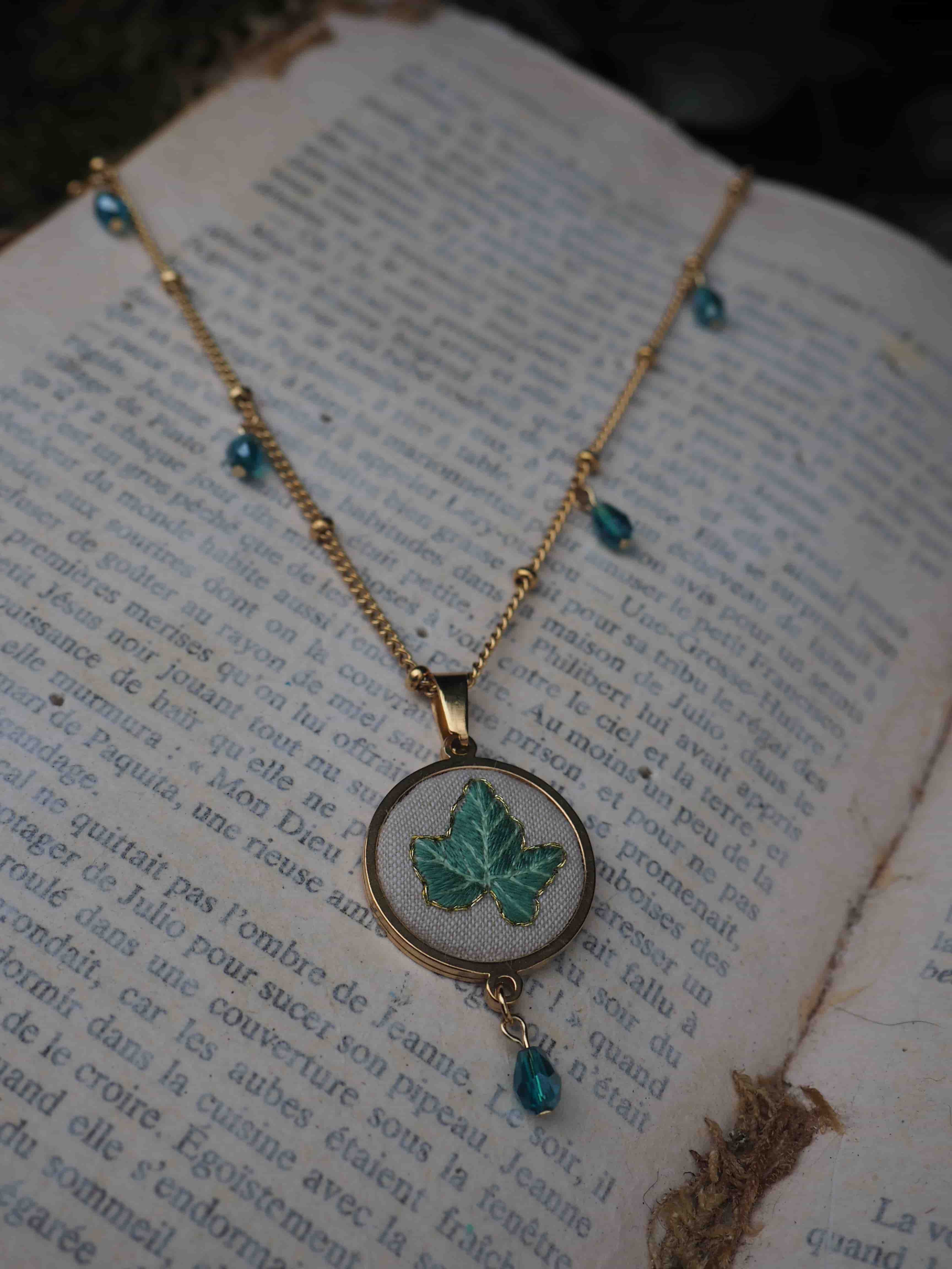 This image features a close-up of a delicate gold-toned necklace resting on an open book with aged, yellowed pages. The necklace has a thin chain adorned with small, evenly spaced gold beads and several small, faceted teal or dark blue beads that dangle at intervals along its length. The central pendant is oval-shaped, with a gold-toned frame. Inside the frame, against a light, possibly white or cream-colored fabric background, is an embroidered or painted design of a single ivy leaf in shades of green. A single matching teal/dark blue faceted bead dangles from the bottom of the oval pendant. The book's pages are filled with printed text in French, suggesting an old novel or literary work. The lighting is soft, highlighting the necklace and the texture of the paper, with a slightly darker, out-of-focus background that hints at natural elements like foliage. The overall aesthetic is vintage, natural, and somewhat whimsical.