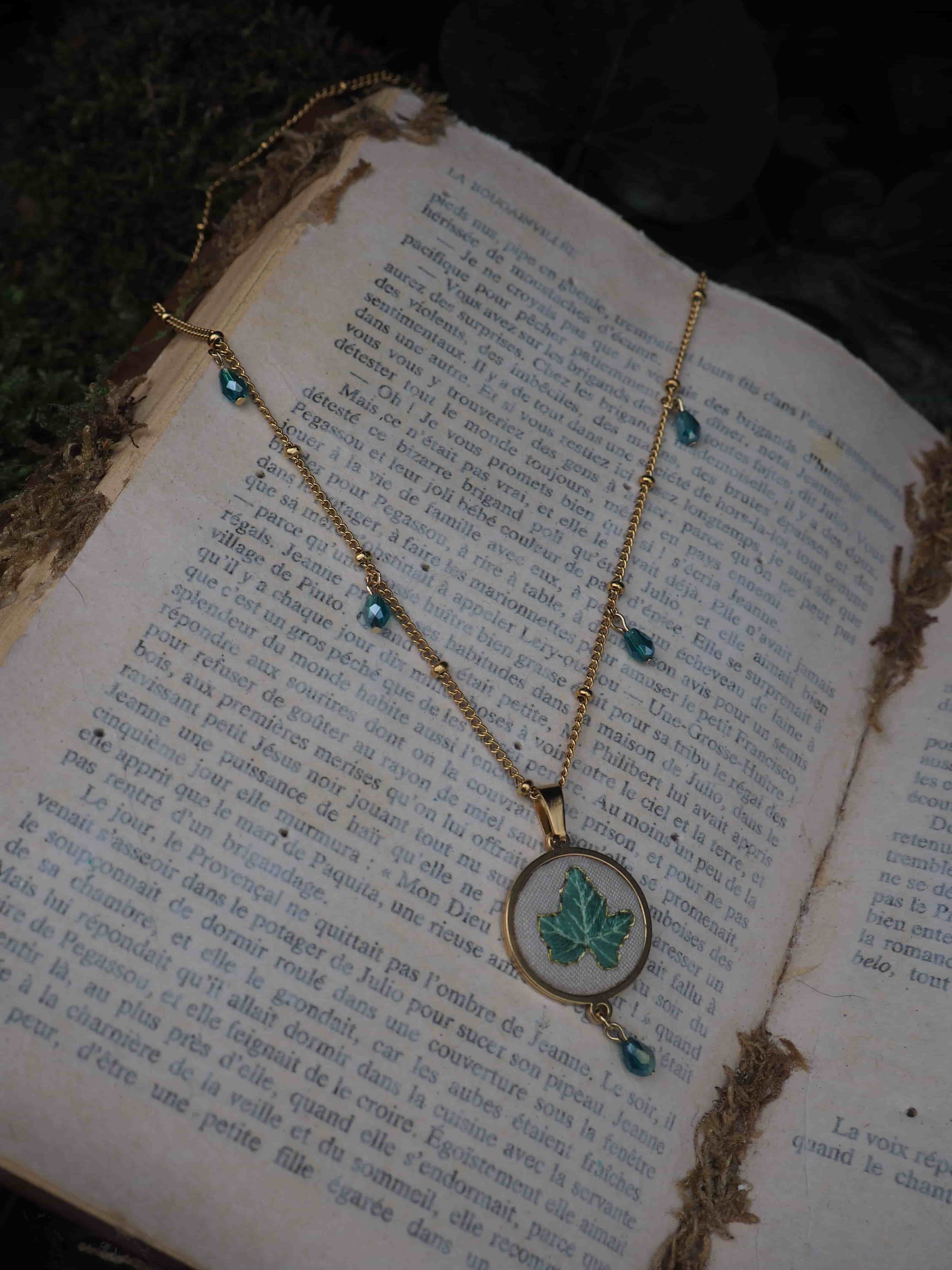 This image features a gold necklace with a pendant and blue beads, resting on an open book. The necklace is made of a delicate gold chain with small gold beads interspersed along its length. Several small, faceted blue beads are attached to the chain at intervals, adding a touch of color. The central pendant is circular, with a gold frame, and features a light green leaf design on a white background. Another blue bead dangles from the bottom of the pendant. The necklace is laid out on an open book with aged, yellowed pages. The book is filled with text printed in black ink, suggesting an old or vintage publication. The pages appear somewhat worn, with slight discoloration and perhaps some minor fraying at the edges. The background of the image is softly blurred, showing hints of dark green foliage or moss, which contributes to a natural, slightly rustic, or antique aesthetic. The lighting appears soft, highlighting the necklace and the book.