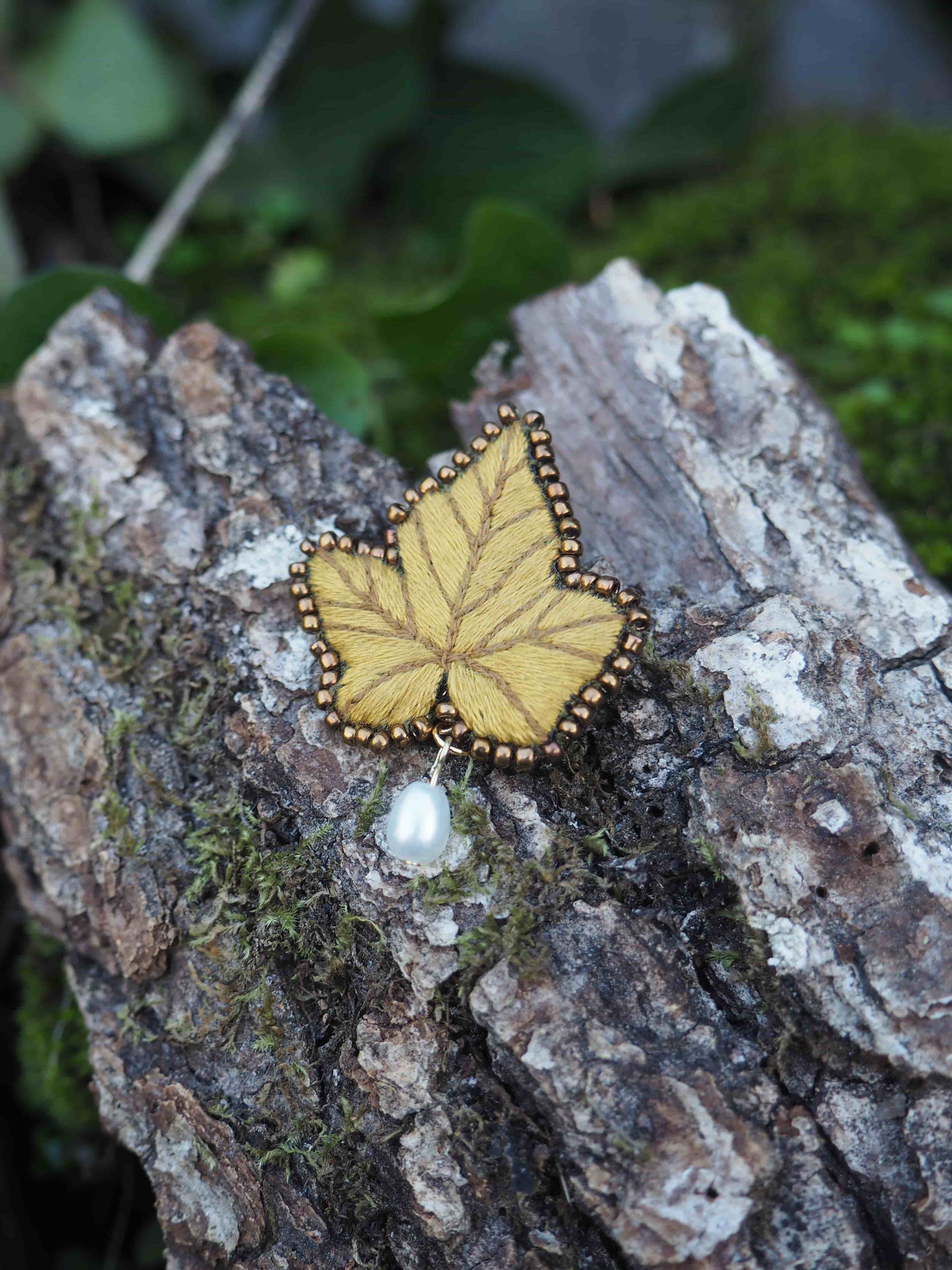 L'image présente une broche en forme de feuille de lierre, posée sur un morceau de bois brut recouvert de mousse. La broche est de couleur jaune moutarde ou ocre clair, avec des nervures de feuille brodées en un fil plus foncé, créant un effet de texture et de profondeur. Le contour de la feuille est orné de petites perles dorées ou cuivrées, ajoutant une touche d'éclat. Sous la broche, suspendue à une petite attache, se trouve une perle de culture blanche, de forme irrégulière, qui pend délicatement. Le morceau de bois sur lequel repose la broche est rugueux et texturé, avec des crevasses et des zones plus claires et plus foncées, typiques de l'écorce. De la mousse verte, de différentes nuances, est visible sur le bois, particulièrement sur le côté gauche et en arrière-plan, suggérant un environnement naturel et forestier. L'éclairage semble naturel, mettant en valeur les détails de la broche et la texture du bois. La profondeur de champ est faible, ce qui permet de bien distinguer la broche et le bois au premier plan, tandis que l'arrière-plan est légèrement flou, avec des touches de vert et de brun. L'ensemble de l'image dégage une atmosphère douce et naturelle, mettant en valeur l'artisanat de la broche dans un cadre organique.