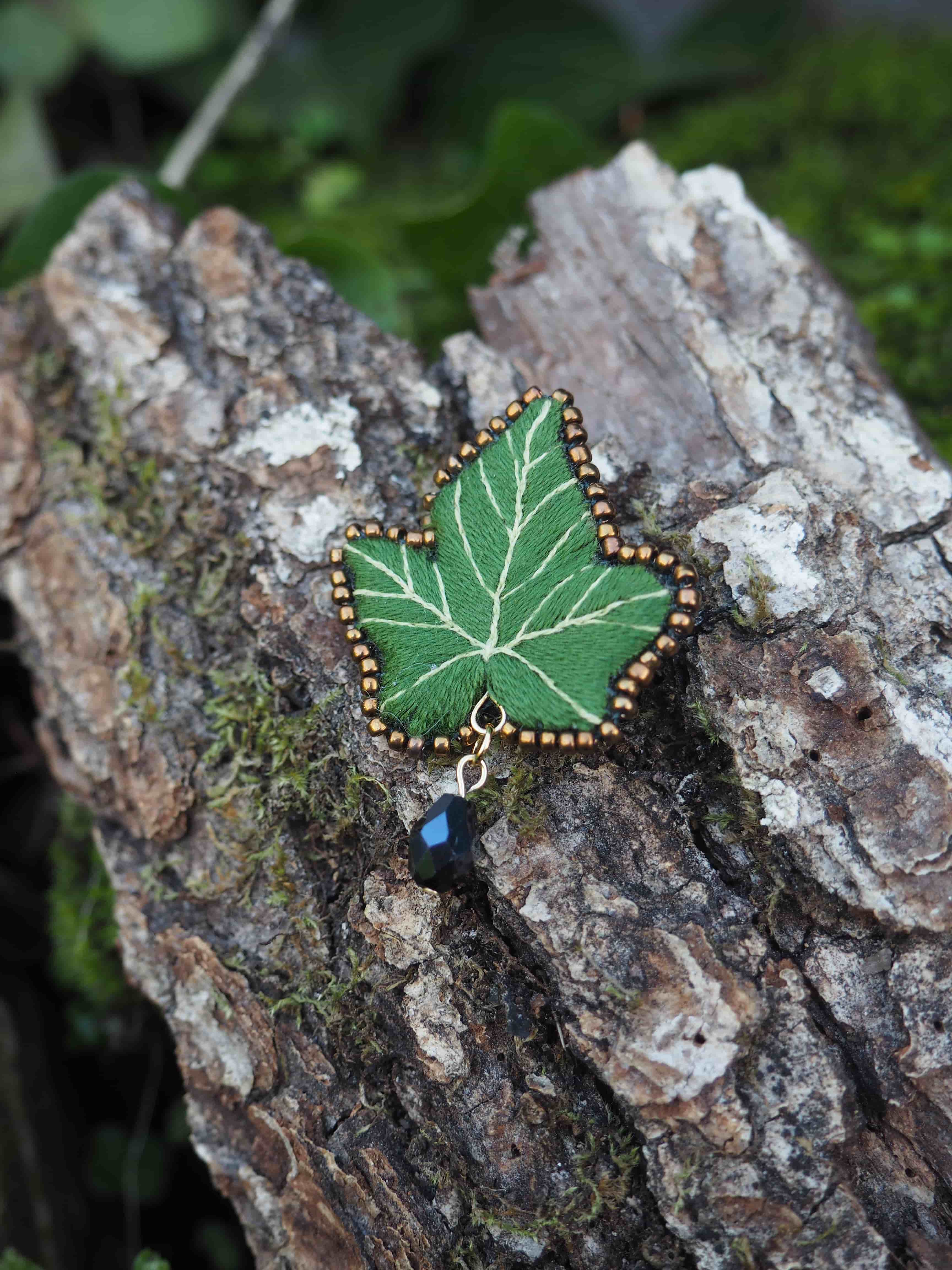 L'image présente une broche en forme de feuille de lierre, posée sur un morceau d'écorce d'arbre. **Description de la broche :**
*   La broche est de couleur verte, imitant la teinte d'une feuille de lierre.
*   Elle est brodée avec des fils plus clairs pour représenter les nervures de la feuille, ajoutant du réalisme et du détail.
*   Le contour de la feuille est orné de petites perles dorées ou de points de broderie de couleur or, ce qui lui donne une finition élégante et un léger éclat.
*   Sous la broche, suspendue par un petit anneau doré, se trouve une perle noire ou un petit cristal taillé, qui ajoute un élément de pendeloque et de sophistication. **Contexte et arrière-plan :**
*   La broche est posée sur un morceau d'écorce d'arbre, qui est rugueuse et texturée, avec des nuances de brun et de gris.
*   De la mousse verte est visible par endroits sur l'écorce, suggérant un environnement naturel et humide.
*   L'arrière-plan est flou, mais on peut distinguer des feuillages verts, ce qui renforce l'idée d'une scène en extérieur ou d'un cadre naturel.
*   La lumière semble douce et naturelle, mettant en valeur les détails de la broche sans créer d'ombres dures. L'ensemble de l'image dégage une atmosphère artisanale et naturelle, soulignant la beauté et la délicatesse de la broche faite à la main.