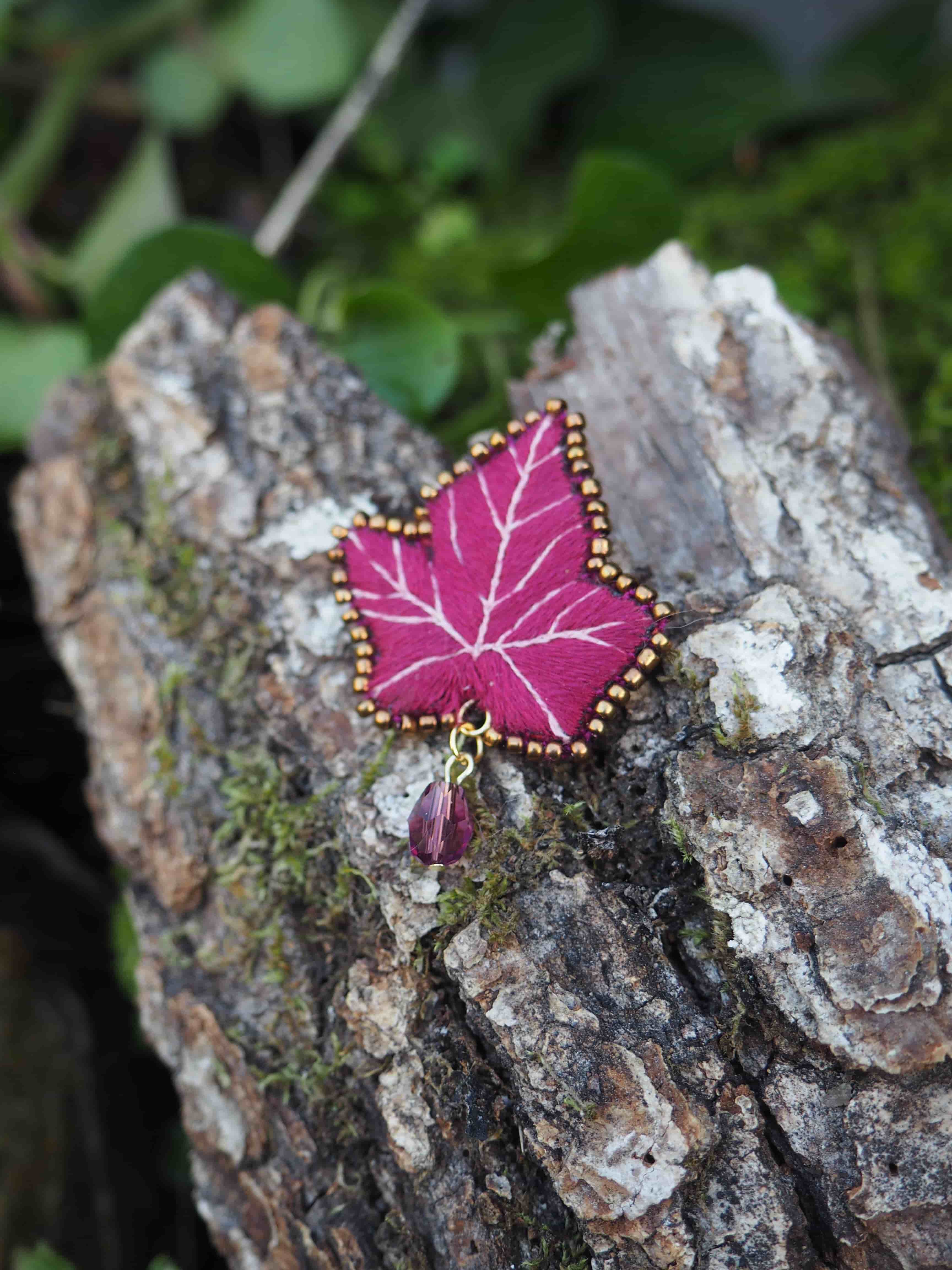Une broche en forme de feuille de lierre, de couleur magenta, est posée sur un morceau de bois. La broche est ornée de perles dorées le long de son contour et d'une petite perle violette suspendue à sa base. Le bois sur lequel elle repose est rugueux et présente des zones de mousse verte. L'arrière-plan est flou, mais on distingue des feuilles vertes, suggérant un environnement naturel. La lumière semble venir d'en haut, éclairant la broche et le bois.