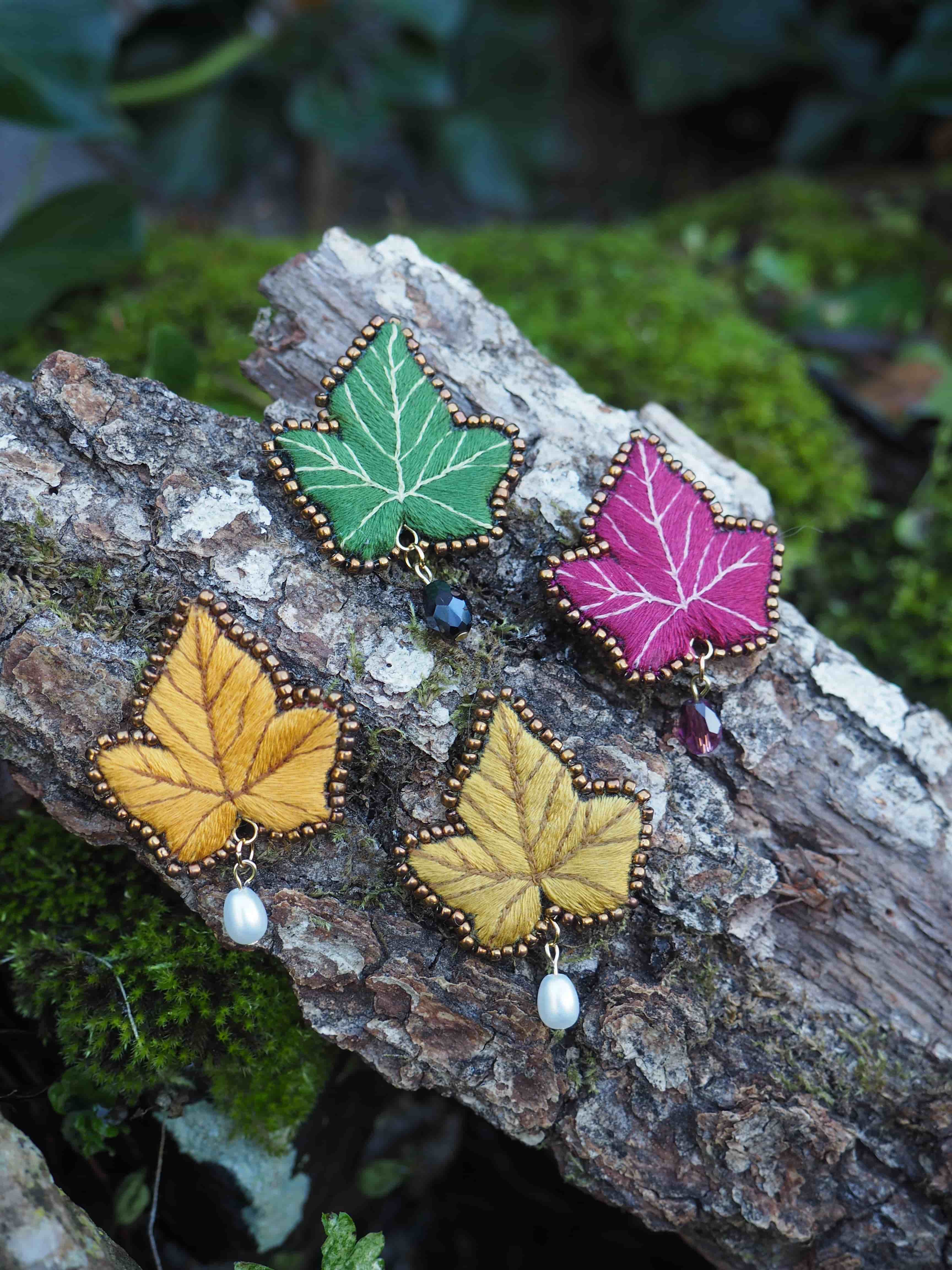 L'image présente quatre broches en forme de feuille, posées sur un morceau de bois recouvert de mousse. Chaque broche est conçue pour ressembler à une feuille de lierre stylisée, avec des détails de nervures brodés et un contour perlé. Voici une description plus détaillée de chaque broche et de l'ensemble : *   **Broche verte :** Située en haut à gauche, elle est d'un vert vif, avec des nervures brodées en fil blanc ou de couleur claire. Le contour est orné de petites perles dorées ou de couleur bronze. Un petit pendentif sombre (peut-être une perle ou une pierre) est attaché à la pointe de la feuille.
*   **Broche fuchsia/magenta :** Placée en haut à droite, cette broche est d'une couleur rose foncé ou fuchsia éclatante. Les nervures sont également brodées en fil clair. Le contour est parsemé de perles dorées ou bronze, et un petit pendentif de couleur foncée (peut-être rouge foncé ou violet) est suspendu à sa pointe.
*   **Broche jaune moutarde/ocre (en bas à gauche) :** Cette broche est d'une teinte jaune moutarde ou ocre. Les nervures sont brodées en fil clair. Le contour est orné de perles dorées ou bronze. Un pendentif en forme de perle de culture blanche et brillante est attaché à sa pointe.
*   **Broche jaune pâle/doré (en bas à droite) :** Similaire à la précédente mais d'une teinte légèrement plus claire, tirant vers le jaune doré ou le beige. Les nervures sont brodées en fil clair. Le contour est également décoré de perles dorées ou bronze. Un pendentif en forme de perle de culture blanche est suspendu à sa pointe. Les broches sont disposées de manière à ce que leurs pointes soient orientées vers le bas, et elles sont légèrement décalées les unes par rapport aux autres. Le fond est un morceau de bois texturé, partiellement recouvert de mousse verte, ce qui crée un contraste naturel et met en valeur les couleurs vives des broches. L'éclairage semble doux et naturel, soulignant les textures des broderies et des perles. L'ensemble dégage une impression d'artisanat délicat et de beauté naturelle.