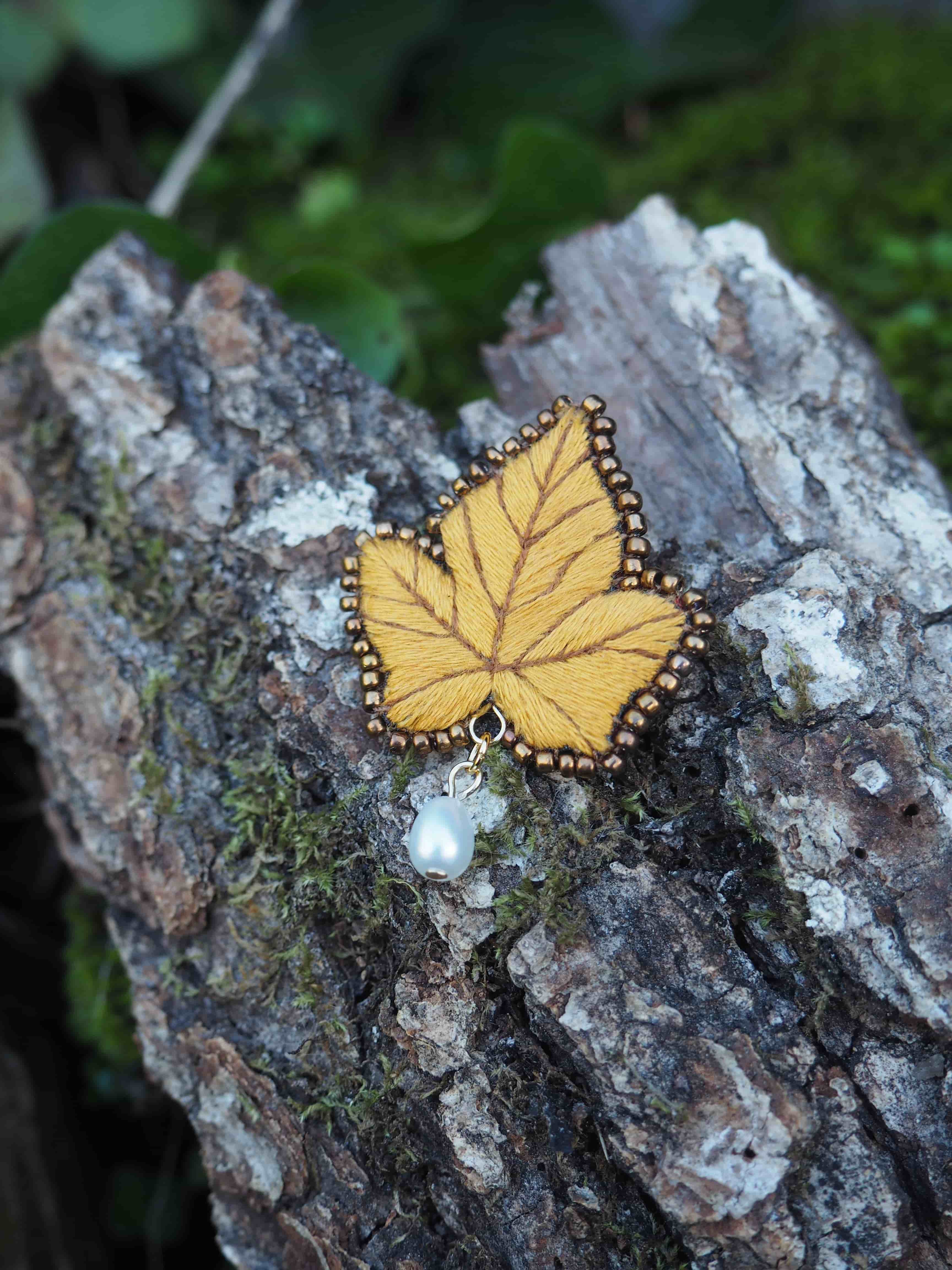 L'image présente une broche en forme de feuille d'érable jaune, posée sur un morceau de bois recouvert de mousse. **Détails de la broche :**
*   **Forme et couleur :** La broche est conçue pour ressembler à une feuille d'érable, avec des lobes distincts et des nervures apparentes. Sa couleur dominante est un jaune moutarde ou ocre, suggérant une feuille d'automne.
*   **Texture et bordure :** La surface de la feuille semble être en tissu ou en feutre, avec des détails brodés pour les nervures. Le contour de la feuille est orné d'une bordure de petites perles dorées ou de couleur bronze, ajoutant une touche d'éclat et de finition.
*   **Pendentif :** Sous la feuille, suspendu à une petite attache dorée, se trouve un petit pendentif. Il est composé d'une perle de forme irrégulière, de couleur blanc nacré ou ivoire, et de quelques petites perles translucides ou de couleur claire, peut-être des perles de verre ou des pierres semi-précieuses. **Contexte et arrière-plan :**
*   **Support :** La broche repose sur un morceau de bois brut, probablement une branche ou un tronc d'arbre, dont l'écorce est texturée et présente des nuances de gris et de brun.
*   **Végétation :** Le bois est partiellement recouvert de mousse verte, ce qui ajoute une touche naturelle et organique à la scène.
*   **Profondeur de champ :** L'arrière-plan est flou, ce qui met en valeur la broche et le morceau de bois au premier plan. On distingue des formes vertes et sombres, suggérant un environnement végétal, peut-être une forêt ou un jardin. **Ambiance générale :**
L'image dégage une atmosphère douce et naturelle, mettant en avant un objet artisanal délicat dans un cadre rustique. La lumière semble naturelle et diffuse, soulignant les textures et les couleurs de la broche et de son environnement.