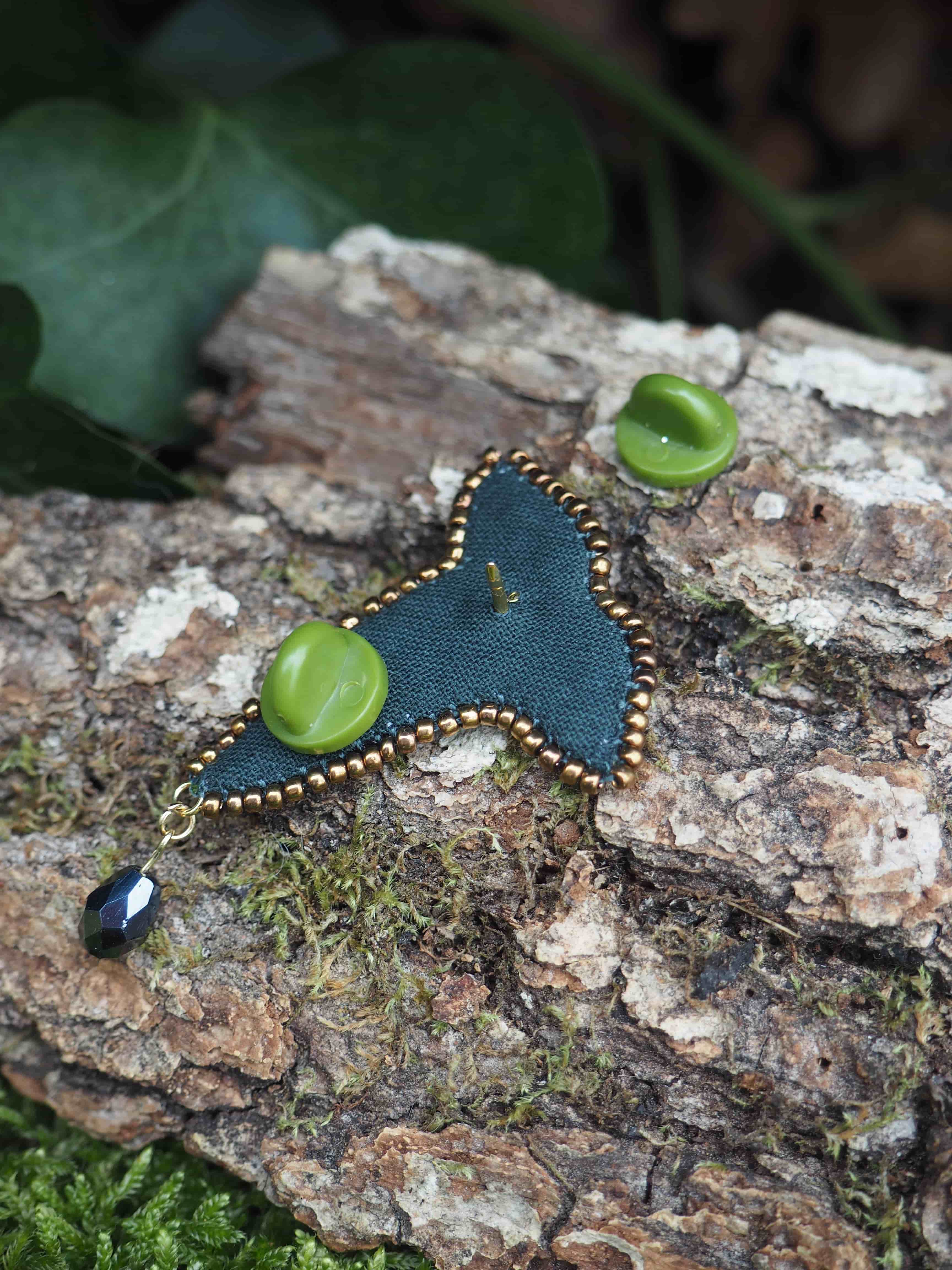 L'image présente une broche en forme de feuille de lierre, posée sur un morceau d'écorce d'arbre texturée, avec de la mousse verte visible en arrière-plan et au premier plan. La broche est de couleur vert foncé, avec des bords ornés de petites perles dorées. Au centre de la broche, il y a une perle ovale ou en forme de goutte de couleur vert clair, qui contraste avec le vert plus sombre du tissu. Une petite épingle dorée est visible au dos de la broche, indiquant son mécanisme de fixation. Accroché à la pointe inférieure de la broche, il y a un petit pendentif composé d'une perle noire ou bleu foncé, de forme allongée et facettée, qui ajoute une touche d'élégance. À côté de la broche, légèrement en haut à droite, se trouve un petit fermoir d'épingle de couleur vert clair, qui correspond probablement à la broche. L'éclairage est doux et naturel, mettant en valeur les textures de l'écorce et les détails de la broche. L'arrière-plan est flou, ce qui aide à focaliser l'attention sur la broche et son environnement immédiat. L'ensemble de la scène évoque une ambiance naturelle et artisanale.