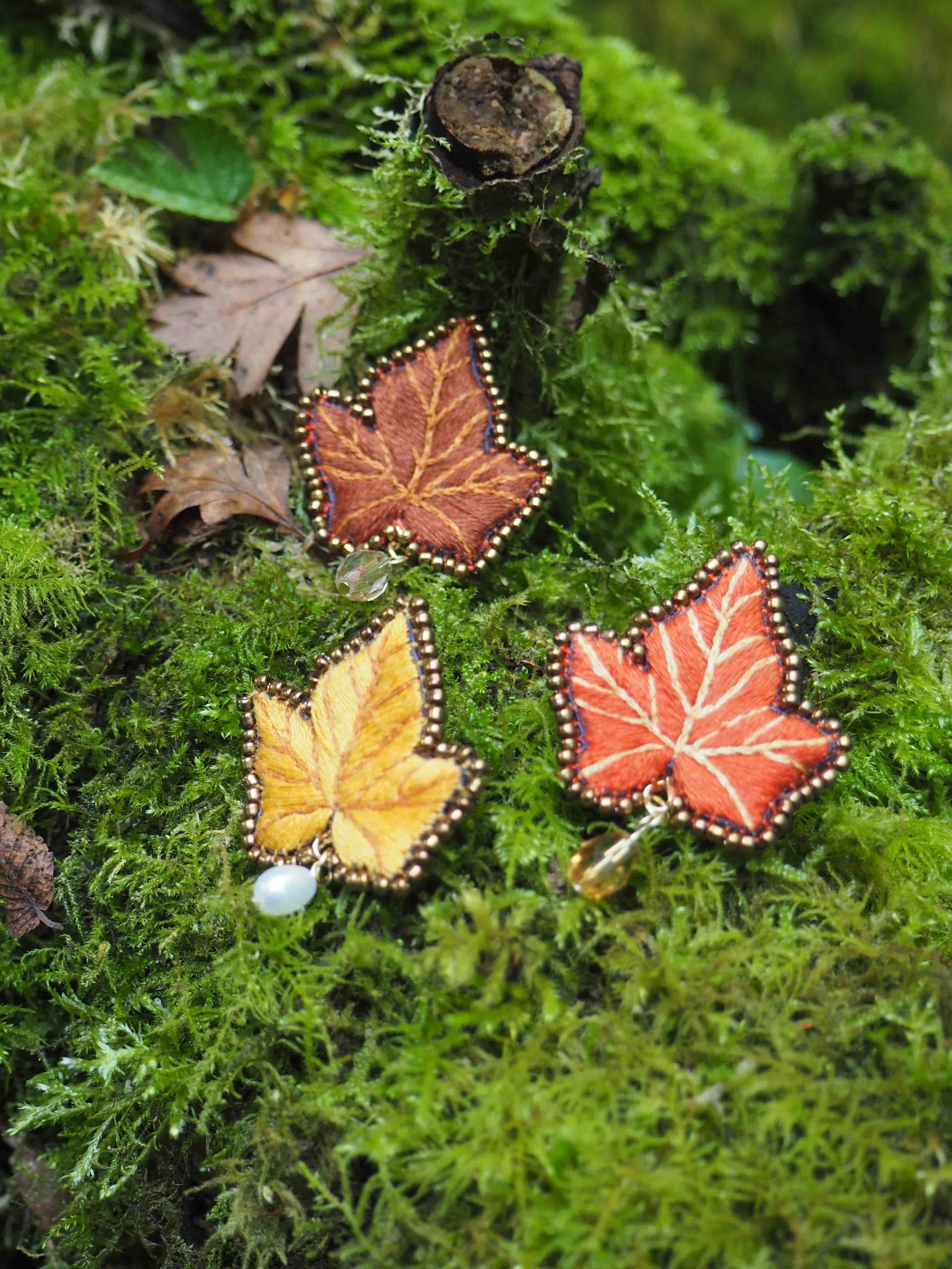 L'image présente trois broches en forme de feuilles d'automne, posées sur un lit de mousse verte luxuriante. Chaque broche est méticuleusement brodée, reproduisant les nervures et les contours d'une feuille. Les couleurs des broches varient, évoquant les teintes automnales :
*   Une broche est d'un orange-brun profond, avec des détails brodés en fil plus clair pour les nervures.
*   Une autre est d'un jaune vif, également avec des nervures brodées. Elle est ornée d'une petite perle blanche nacrée suspendue à sa base.
*   La troisième broche est d'un orange plus clair, presque rouge, avec des nervures blanches contrastantes. Elle est agrémentée d'une petite perle ou d'une goutte de verre de couleur ambre suspendue. Les bords de chaque broche sont ornés de petites perles dorées ou de rocailles, ajoutant une touche de finition délicate. L'arrière-plan est dominé par une mousse verte dense et texturée, avec quelques feuilles d'automne naturelles et des brindilles visibles, ce qui renforce le thème naturel et automnal de la scène. La lumière semble douce, mettant en valeur les détails et les textures des broches et de la mousse.