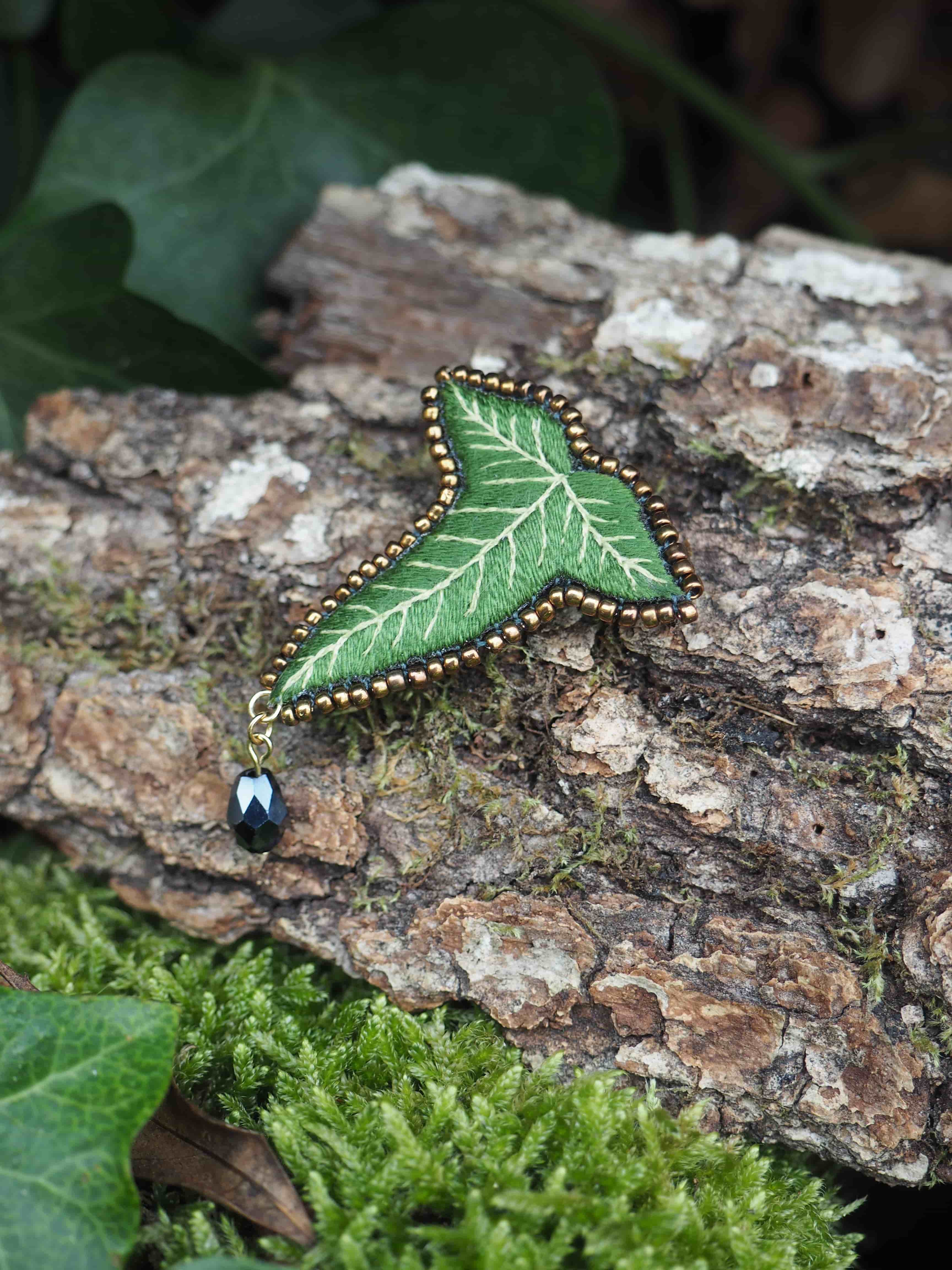 L'image présente une broche en forme de feuille de lierre, posée sur un morceau d'écorce d'arbre texturée et recouverte de mousse verte. La broche est le point focal de l'image. Elle est de couleur verte, avec des nervures plus claires brodées qui imitent celles d'une vraie feuille de lierre. Le contour de la broche est orné de petites perles dorées, ajoutant une touche d'éclat. Une petite perle noire brillante est suspendue à la pointe inférieure de la broche par un anneau doré, agissant comme un petit pendentif. L'arrière-plan est composé d'éléments naturels. Le morceau d'écorce sur lequel repose la broche est rugueux et présente des nuances de brun et de gris, avec des taches de mousse verte plus foncée. En dessous et autour de l'écorce, on peut voir une couche dense de mousse verte vive, créant un contraste de texture et de couleur. Quelques feuilles de lierre naturelles, de couleur vert foncé, sont visibles dans le coin inférieur gauche et en haut à gauche de l'image, renforçant le thème naturel et végétal. La lumière semble naturelle et douce, mettant en valeur les détails de la broche et les textures de l'environnement. L'ensemble de l'image dégage une atmosphère naturelle et délicate, soulignant le caractère artisanal et la beauté de la broche.