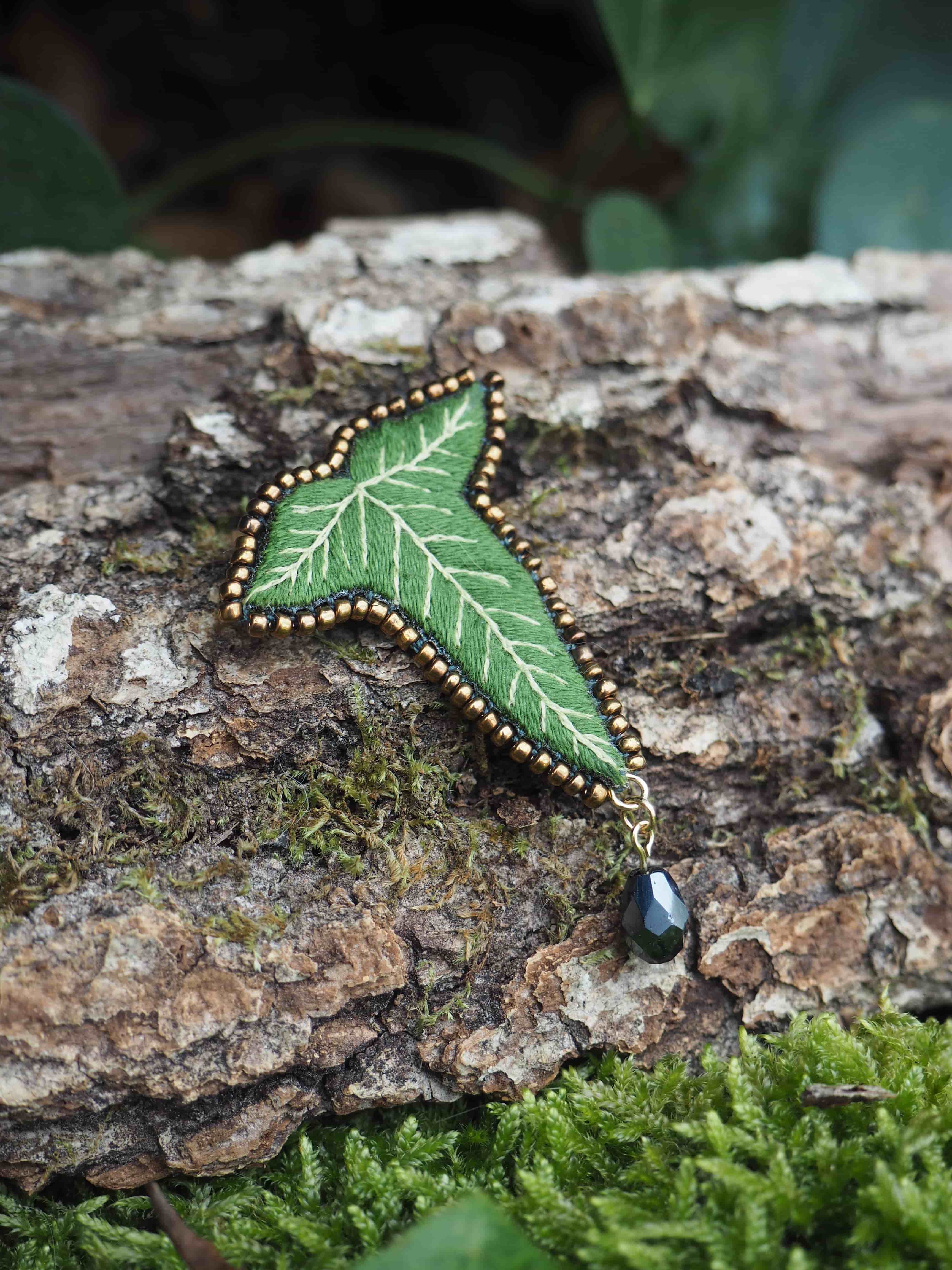 L'image présente une broche en forme de feuille de lierre, posée sur un morceau de bois recouvert de mousse. La broche est de couleur verte, avec des nervures plus claires brodées pour imiter l'aspect naturel d'une feuille. Le contour de la feuille est orné de petites perles dorées, ajoutant une touche d'éclat. À l'extrémité inférieure de la broche, une petite perle noire en forme de goutte est suspendue, apportant un contraste et un mouvement subtil. Le fond est composé d'un tronc d'arbre ou d'une branche épaisse, dont l'écorce est texturée et de couleur marron. De la mousse verte, de différentes nuances, recouvre partiellement le bois, créant un environnement naturel et doux. La profondeur de champ est faible, ce qui met en valeur la broche au premier plan, tandis que l'arrière-plan est légèrement flou. L'éclairage semble naturel, mettant en évidence les détails de la broche et les textures du bois et de la mousse. L'ensemble de l'image dégage une atmosphère calme et naturelle, soulignant le caractère artisanal et délicat de la broche.