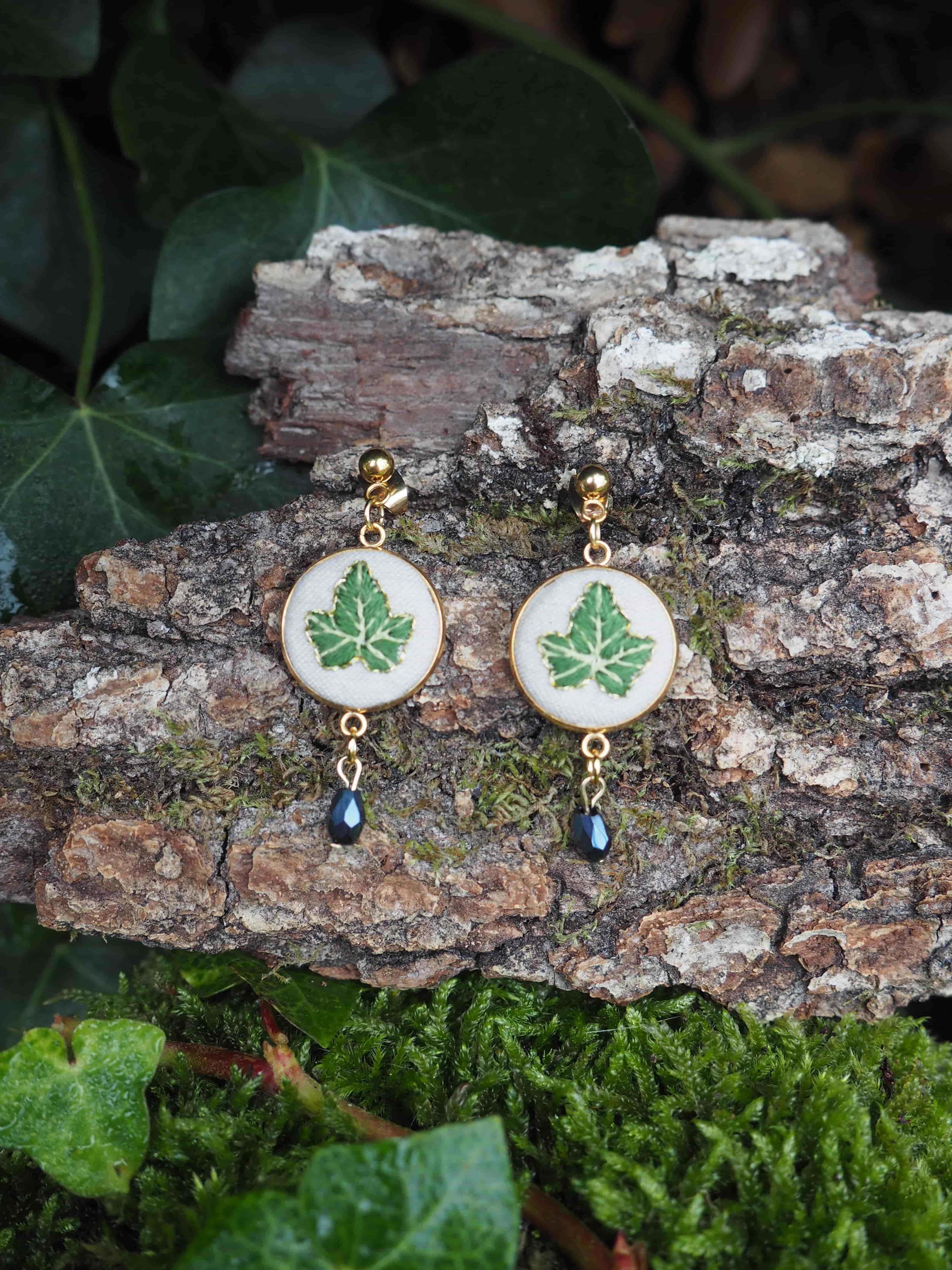 This close-up shot features a pair of nature-inspired earrings resting on a piece of tree bark, surrounded by lush green foliage. The earrings are identical, each consisting of a gold-colored stud from which an oval-shaped pendant hangs. The pendant has a gold-colored rim and a white background, upon which a detailed green leaf design is depicted. Below the oval pendant, a small, dark, teardrop-shaped bead dangles, adding a subtle touch of elegance. The tree bark beneath the earrings is textured, with visible grooves and patches of green moss, suggesting an outdoor or natural setting. In the foreground and background, various green leaves and moss create a soft, organic backdrop, enhancing the natural theme of the jewelry. The lighting appears soft, highlighting the details of the earrings and their surroundings.
