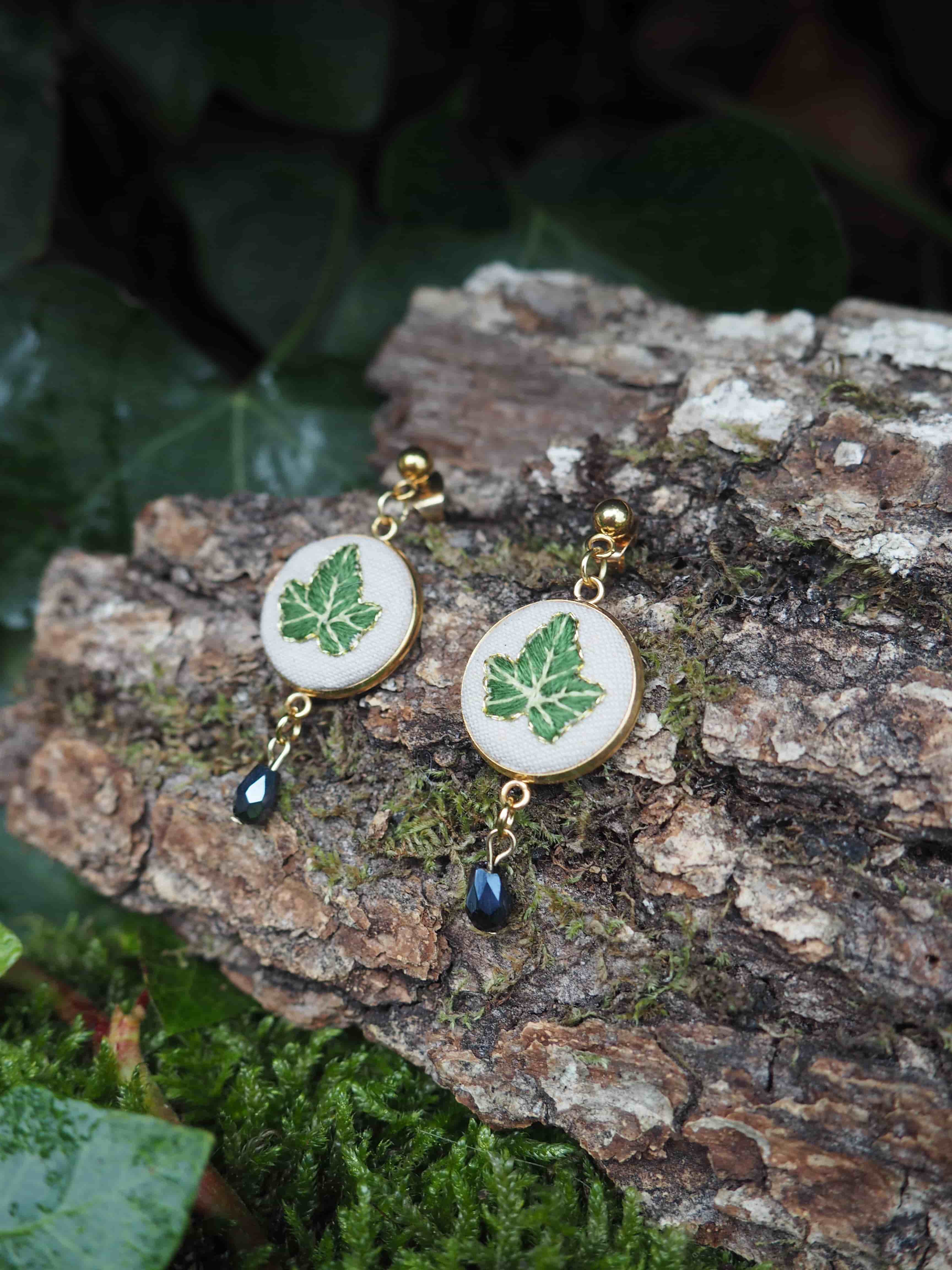 This image features a pair of handcrafted earrings, each adorned with a delicate green leaf design, set against a natural, rustic backdrop. Here's a breakdown of the visual properties: **Subject:**
*   **Earrings:** There are two earrings, appearing to be a matching pair.
    *   **Design:** Each earring consists of a circular, flat component with a white or off-white background. Centered on this background is a detailed green leaf, possibly an ivy leaf, with visible veins. The leaf design has a slightly raised or embroidered appearance.
    *   **Frame:** The circular component is framed by a thin, golden-colored metal bezel.
    *   **Dangle:** Attached to the bottom of each circular component is a small, dark, faceted bead (possibly glass or crystal) that dangles.
    *   **Hook/Stud:** At the top of each earring, there's a small, golden-colored ball, suggesting they are stud earrings or have a stud-like attachment.
    *   **Material:** The golden elements appear to be metal, possibly brass or gold-plated. The white background could be fabric, resin, or enamel. The green leaf looks like a miniature embroidery or a detailed print under a clear coating. **Setting/Background:**
*   **Natural Elements:** The earrings are displayed on a piece of rough, textured tree bark, which is covered in patches of vibrant green moss.
*   **Foliage:** In the background, out of focus, are dark green leaves, likely ivy, contributing to a natural, woodland aesthetic.
*   **Color Palette:** The dominant colors are greens (moss, leaves, leaf design on earrings), browns (bark), and gold (earring metal), with touches of white/cream and dark blue/black from the dangling beads. **Composition and Lighting:**
*   **Arrangement:** The earrings are placed side-by-side on the bark, slightly angled, allowing both to be clearly visible.
*   **Depth of Field:** The foreground (earrings and bark) is in sharp focus, while the background foliage is softly blurred, creating a sense of depth and drawing attention to the jewelry.
*   **Lighting:** The lighting appears natural and soft, possibly overcast or diffused, highlighting the details of the earrings without harsh shadows. **Overall Impression:**
The image conveys a sense of natural beauty, craftsmanship, and elegance. The choice of a natural setting complements the leaf motif of the earrings, suggesting an organic or botanical theme. The focus is clearly on showcasing the intricate details of the handmade jewelry.