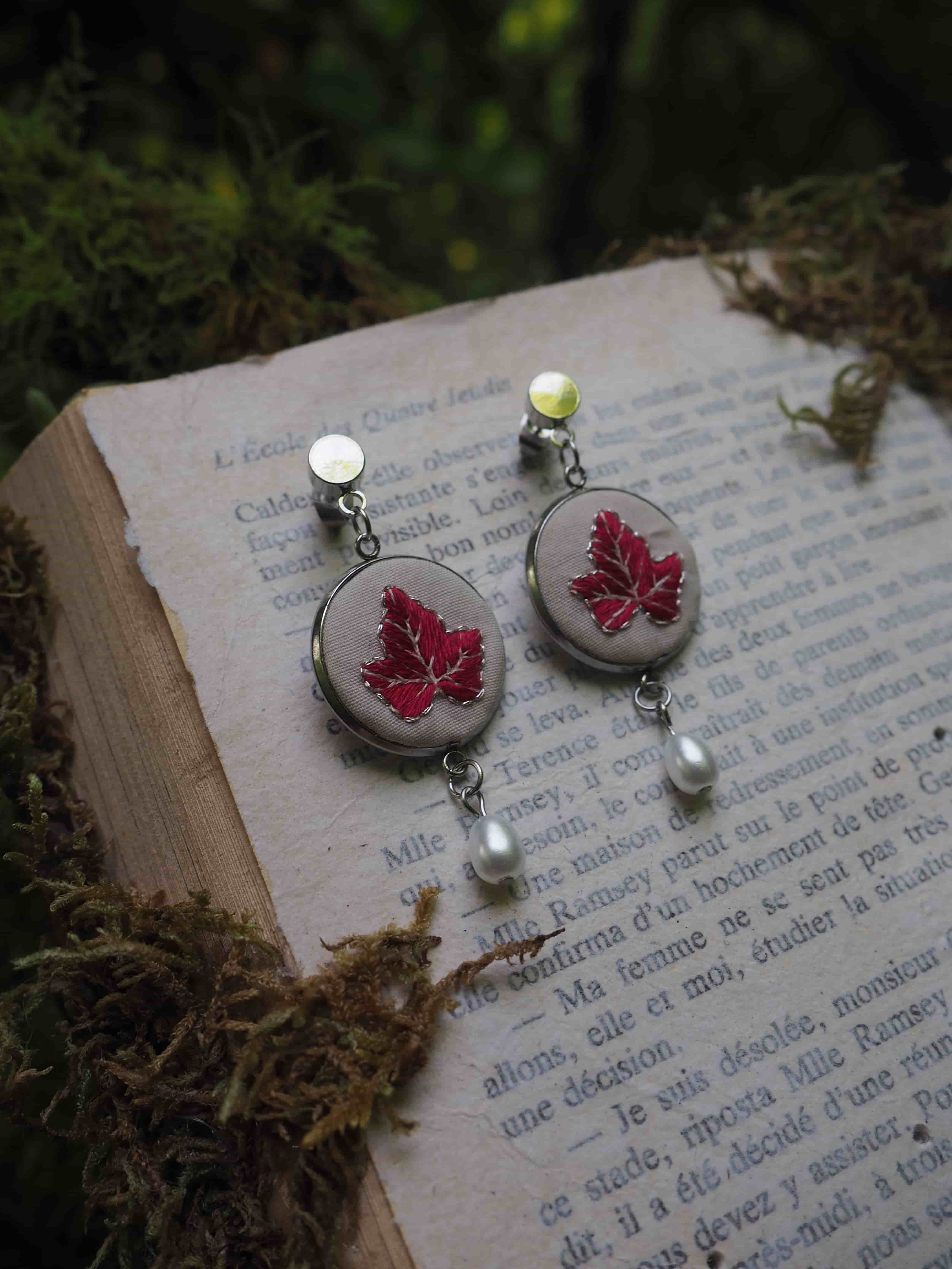 Cette image montre une paire de boucles d'oreilles artisanales posées sur un livre ouvert dans un cadre naturel. Les boucles d'oreilles sont très élégantes, avec des médaillons argentés contenant chacun une feuille d'érable rouge pressée. Au bas de chaque boucle d'oreille pend une petite perle blanche. Les bijoux sont photographiés sur les pages d'un livre en français, avec de la mousse ou des éléments végétaux verts flous en arrière-plan, créant une ambiance très poétique et naturelle. Le design est délicat et évoque l'automne avec les feuilles d'érable rouges.
