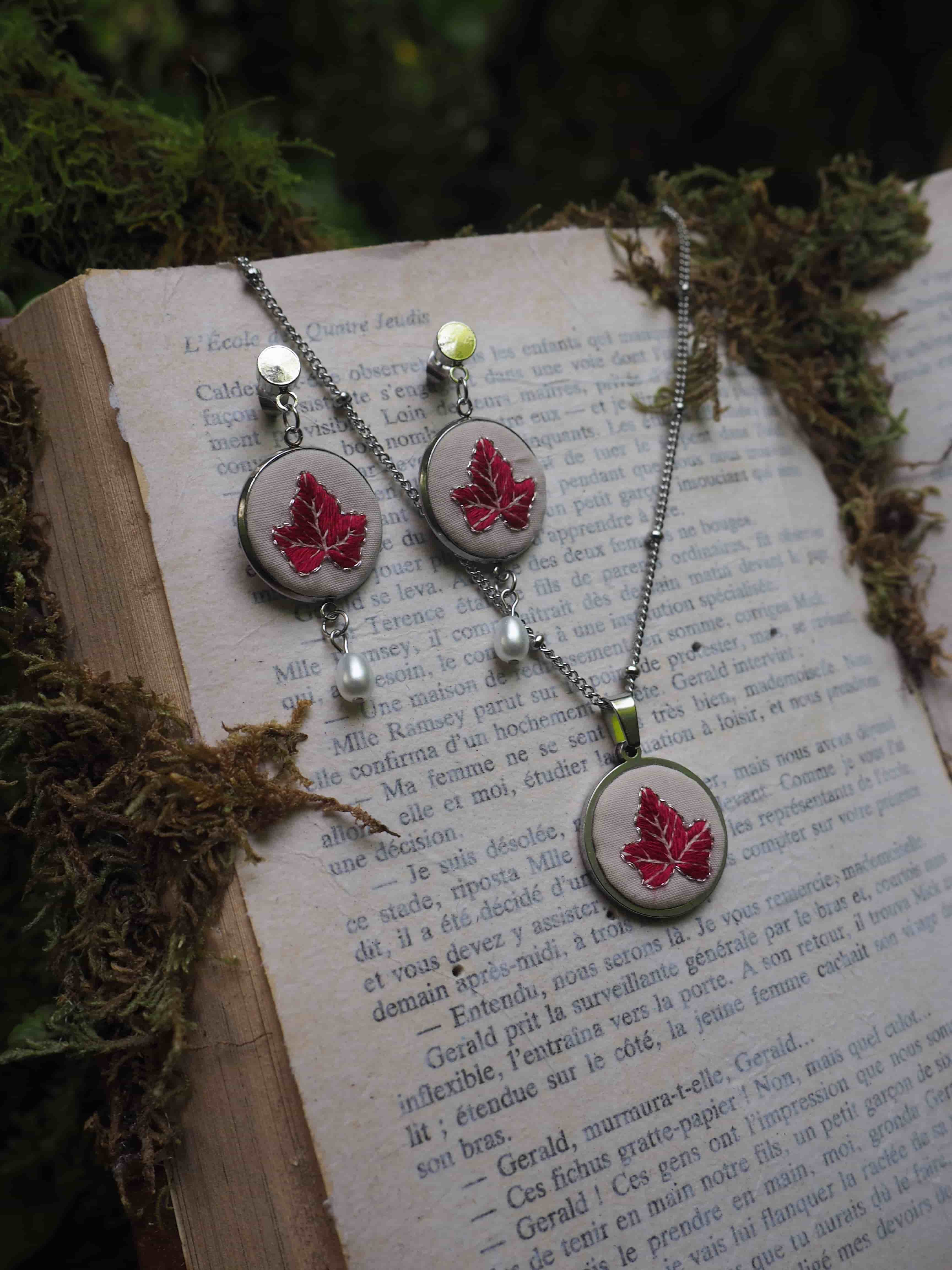 Cette image montre un ensemble de bijoux artisanaux délicats composé de boucles d'oreilles et d'un collier assortis. Chaque pièce présente une feuille d'érable rouge brodée sur un fond blanc, encadrée dans ce qui semble être des supports métalliques circulaires. Les bijoux sont photographiés sur les pages ouvertes d'un livre ancien, avec de la mousse ou des éléments naturels verts disposés autour. Les boucles d'oreilles et le pendentif sont ornés de petites perles blanches et de ce qui semble être des perles de verre vert clair. L'ensemble dégage une atmosphère romantique et naturelle, avec un style vintage délicat.
