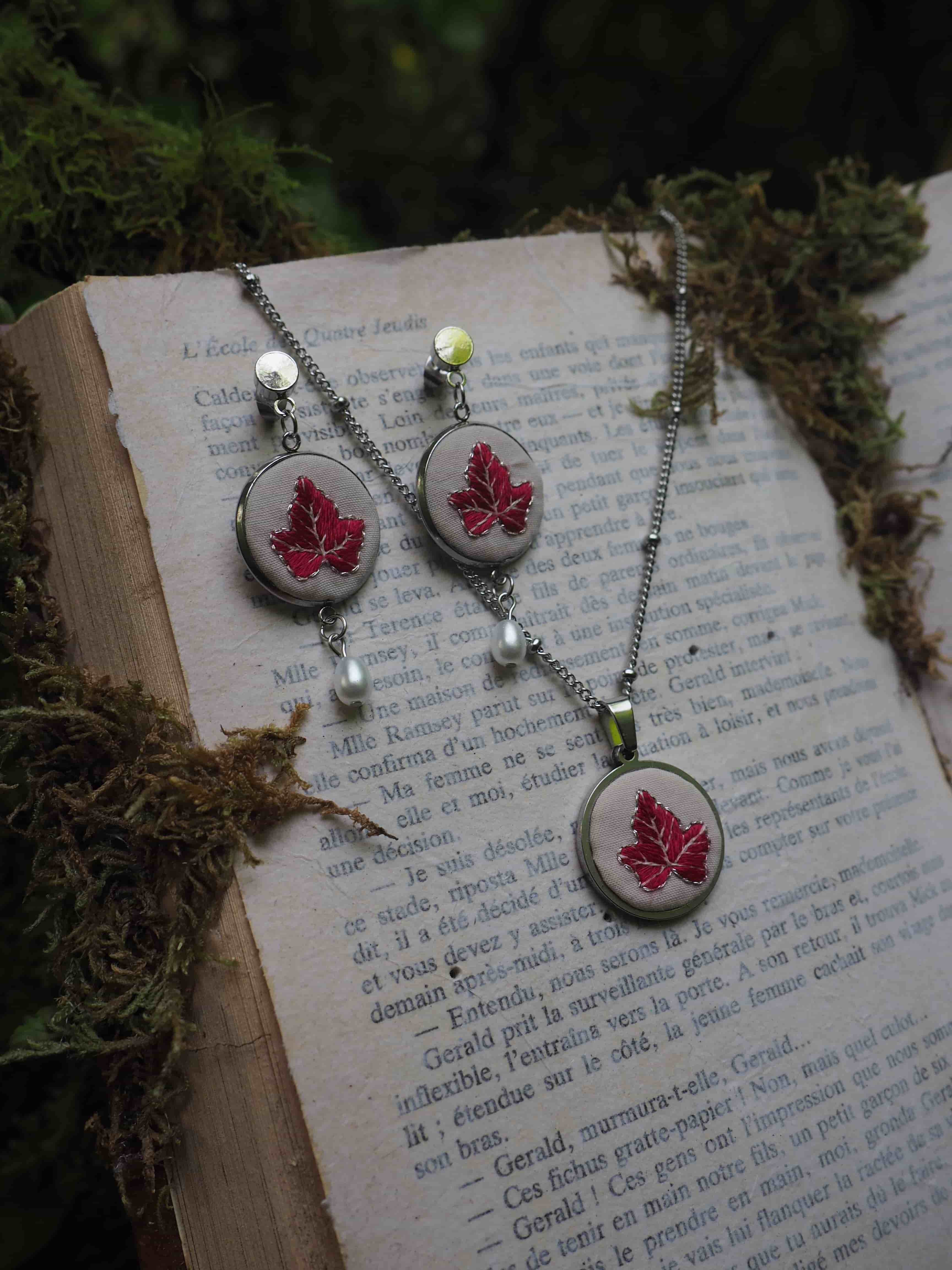 Cette image montre un ensemble de bijoux élégants composé de boucles d'oreilles et d'un collier assortis, disposés sur les pages d'un livre ancien. Les bijoux présentent un motif de feuille d'érable rouge dans des médaillons argentés. Le design est agrémenté de petites perles blanches et de ce qui semble être des perles de verre vert clair. L'arrière-plan montre du texte imprimé sur les pages du livre et quelques éléments de mousse ou de végétation, créant une ambiance naturelle et romantique. La composition est artistique et évoque un style vintage ou fait main.