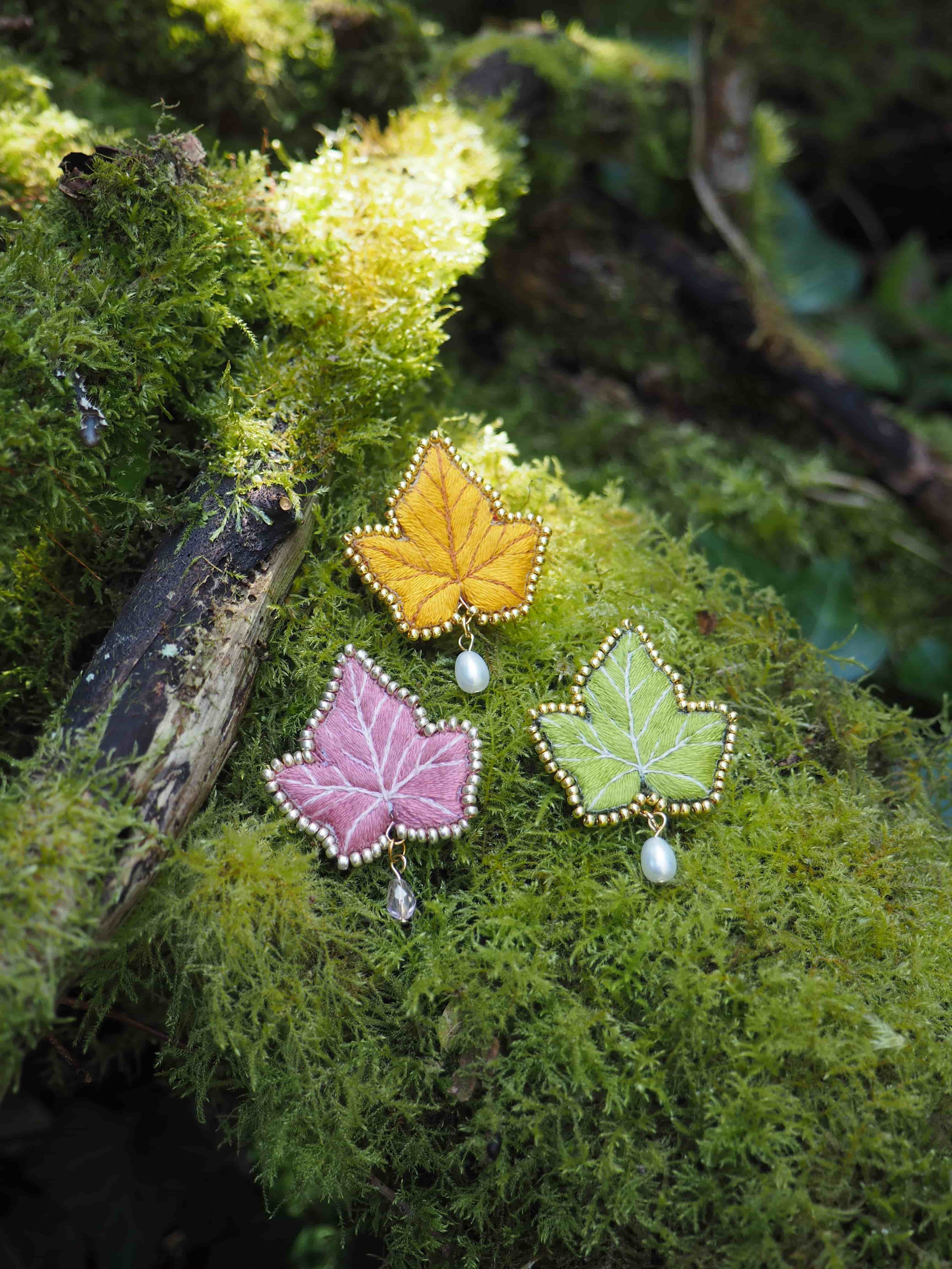 Cette image montre trois jolies broches ou pendentifs en forme de feuilles d'érable, posées sur un lit de mousse verte dans un cadre naturel. Les feuilles sont décorées de manière délicate avec ce qui semble être des perles et des bordures ornementales. Elles sont de différentes couleurs : une jaune/orange, une rose, et une vert clair. Chaque feuille semble avoir une petite perle qui y est attachée. La mousse verte qui sert d'arrière-plan donne un aspect très naturel et féérique à la composition, avec une belle lumière douce qui filtre à travers le feuillage. C'est une photo très artistique qui combine des éléments de bijouterie artisanale avec la beauté de la nature.