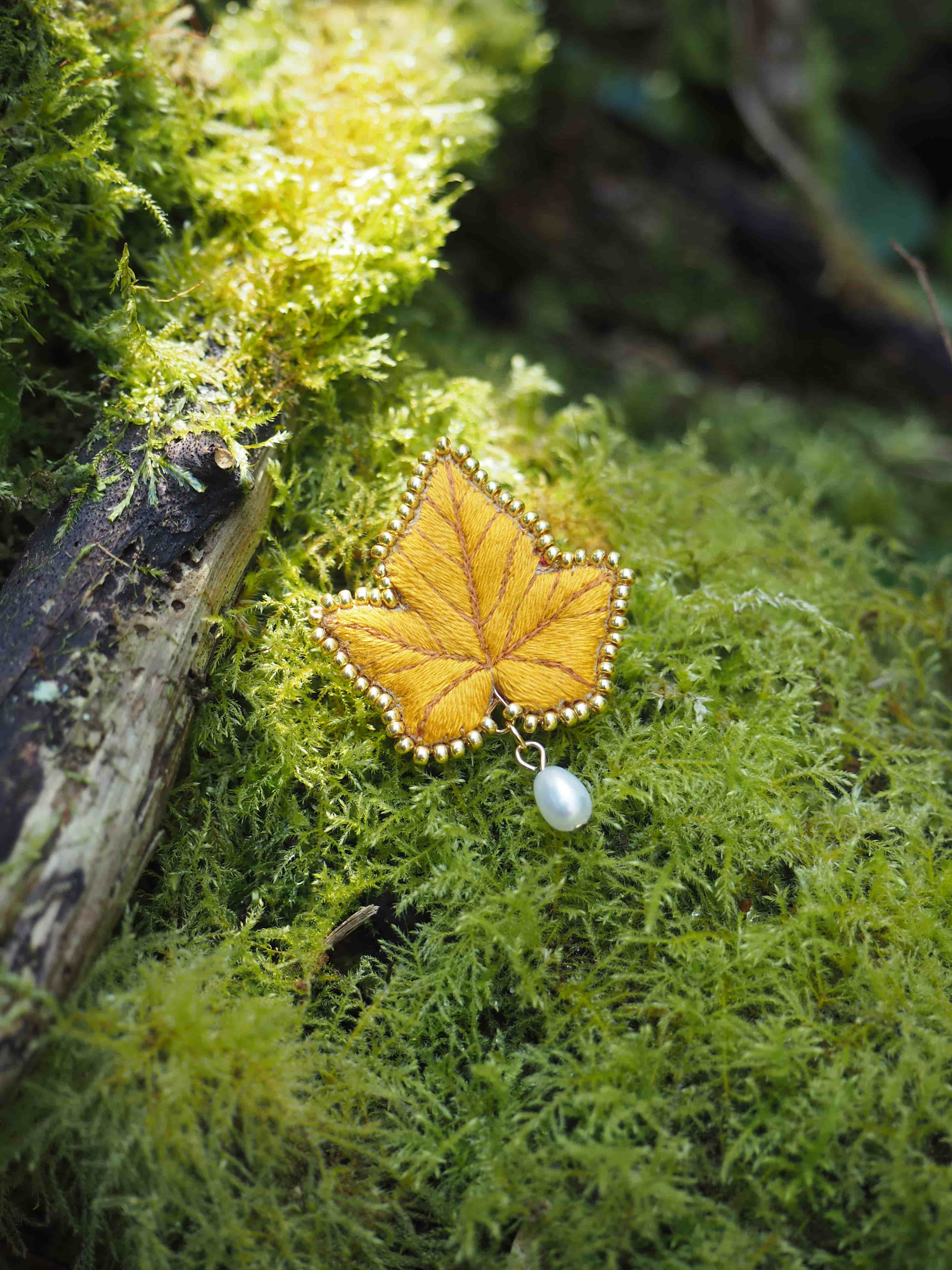 Cette image montre un délicat pendentif ou une broche en forme de feuille d'automne, posé sur un lit de mousse verte luxuriante. Le bijou semble être fait en métal doré ou cuivré, avec des détails finement travaillés qui imitent les nervures naturelles d'une feuille. Une petite perle blanche ou une pierre est suspendue au bas de la feuille. Le contraste entre l'éclat métallique du bijou et la texture douce et naturelle de la mousse crée une composition très poétique et féerique. L'arrière-plan flou et verdoyant ajoute à l'ambiance forestière et mystérieuse de la scène.