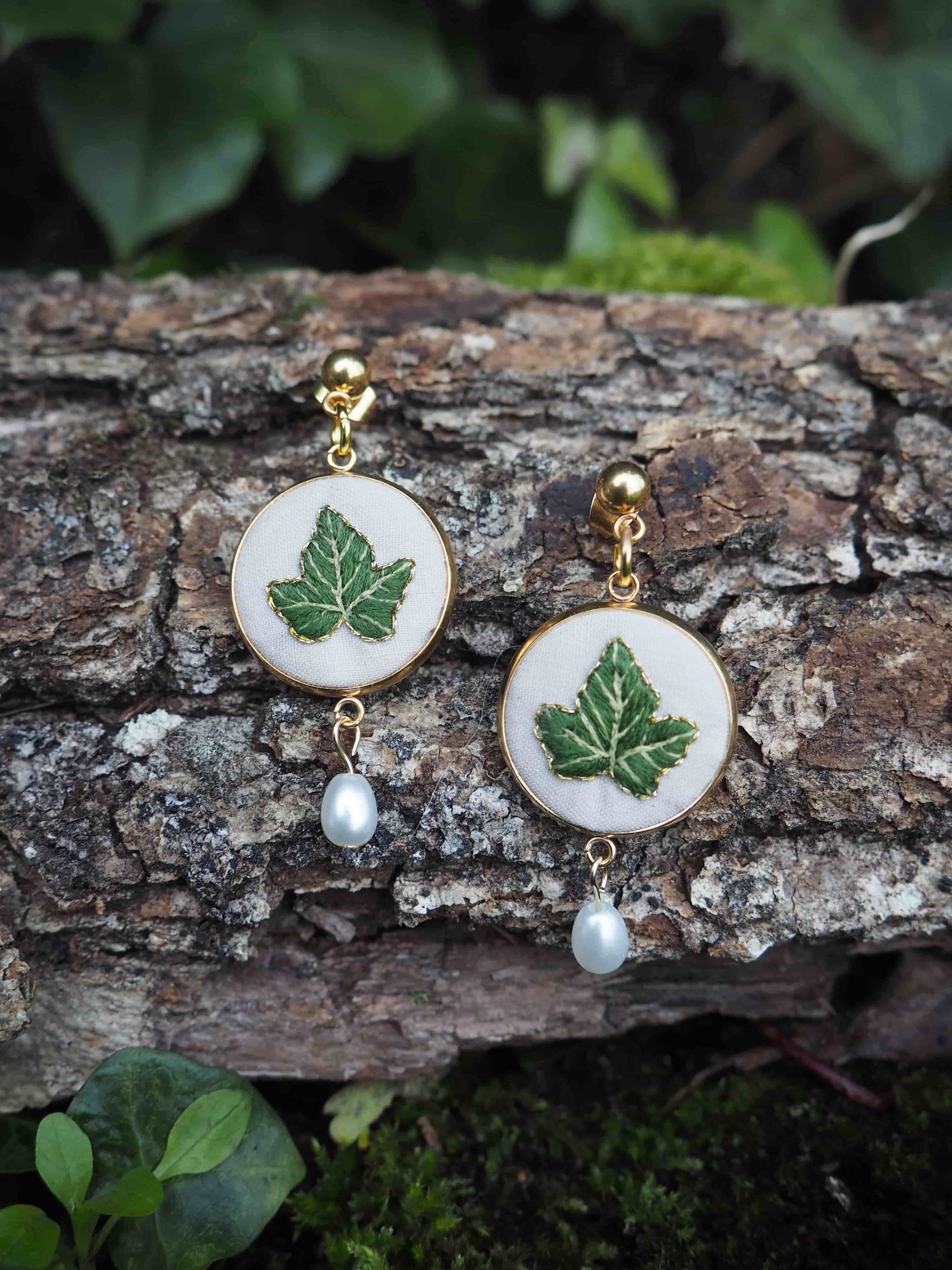 Cette image montre une paire de boucles d'oreilles artisanales élégantes posées sur un morceau d'écorce d'arbre. Les boucles d'oreilles sont composées de médaillons blancs circulaires sur lesquels sont peintes des feuilles d'érable vertes. Chaque boucle d'oreille est ornée d'une petite perle blanche qui pend en dessous du médaillon. Les attaches semblent être en métal doré. L'arrière-plan naturel avec l'écorce texturée et quelques feuilles vertes floues met parfaitement en valeur ces bijoux délicats qui allient nature et élégance.