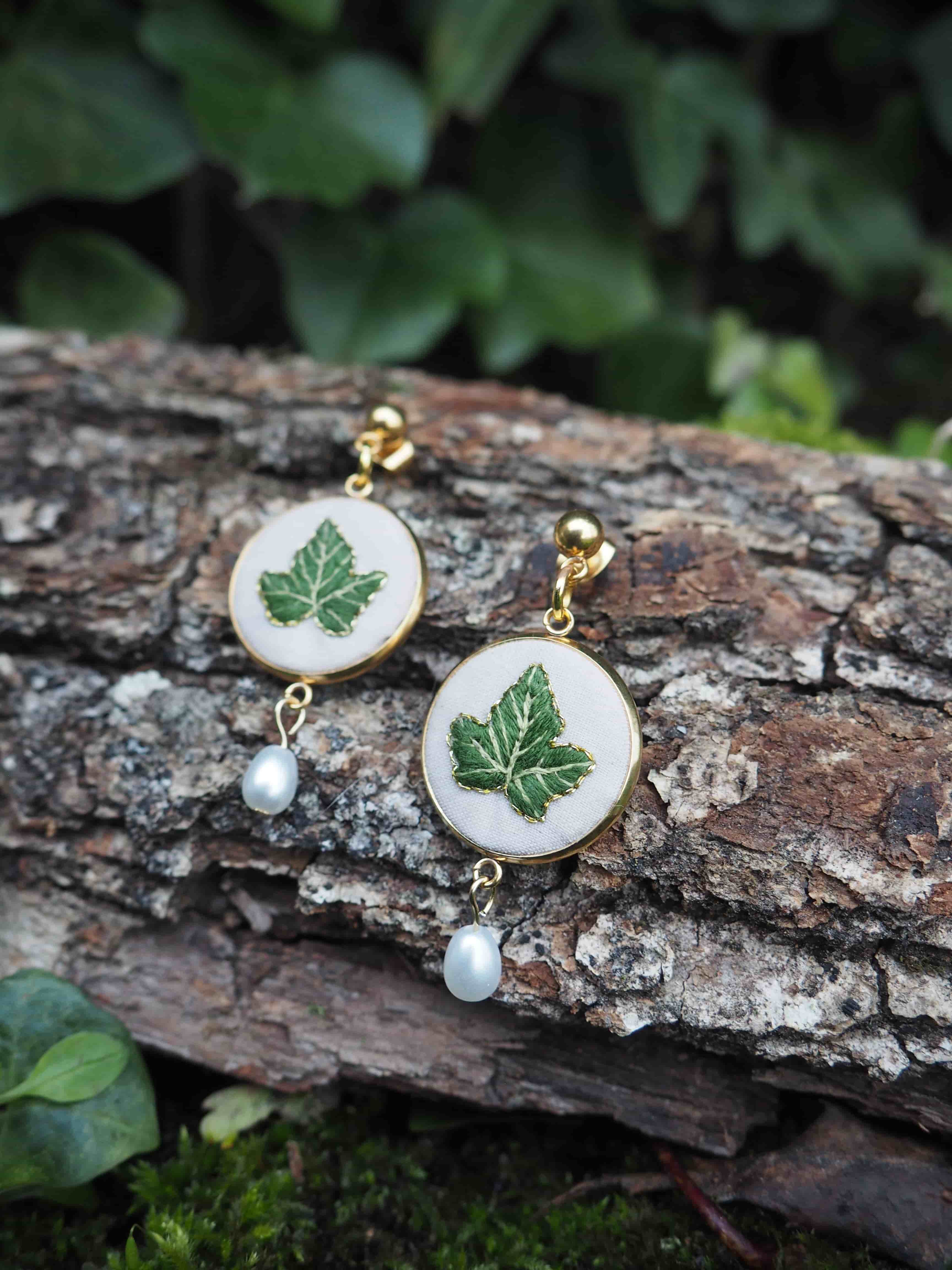 Cette image montre une paire de boucles d'oreilles artisanales délicates posées sur un morceau d'écorce d'arbre. Les boucles d'oreilles sont composées de médaillons blancs circulaires sur lesquels sont peintes des feuilles d'érable vertes. Chaque boucle d'oreille est ornée d'une petite perle blanche qui pend en dessous du médaillon. Les attaches semblent être en métal doré. L'arrière-plan montre du feuillage vert flou, créant une ambiance naturelle et forestière qui complimente parfaitement le motif de feuille d'érable sur les bijoux.