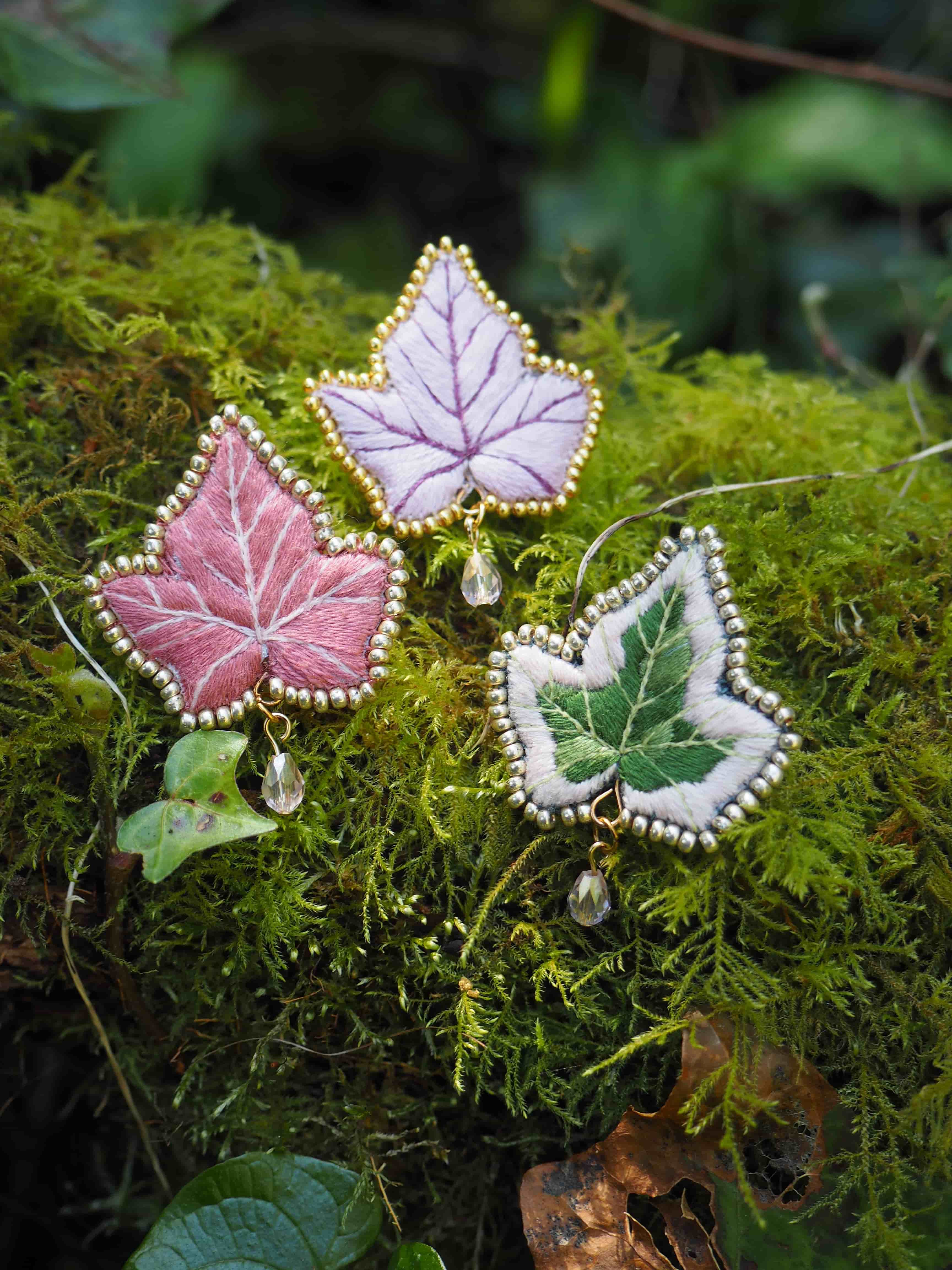 Cette image montre trois jolies broches ou pendentifs en forme de feuilles d'érable, posées sur un lit de mousse verte. Les feuilles sont délicatement confectionnées avec des bordures argentées et sont de trois couleurs différentes : rose, lavande et vert pâle. Chaque feuille semble être ornée d'une petite perle ou cristal suspendu. Le contraste entre les bijoux délicats et la mousse naturelle crée une ambiance féerique et enchantée. L'arrière-plan est flou mais on peut apercevoir d'autres éléments naturels comme des feuilles vertes. C'est une composition très artistique qui mélange élégamment des éléments de joaillerie avec la nature.