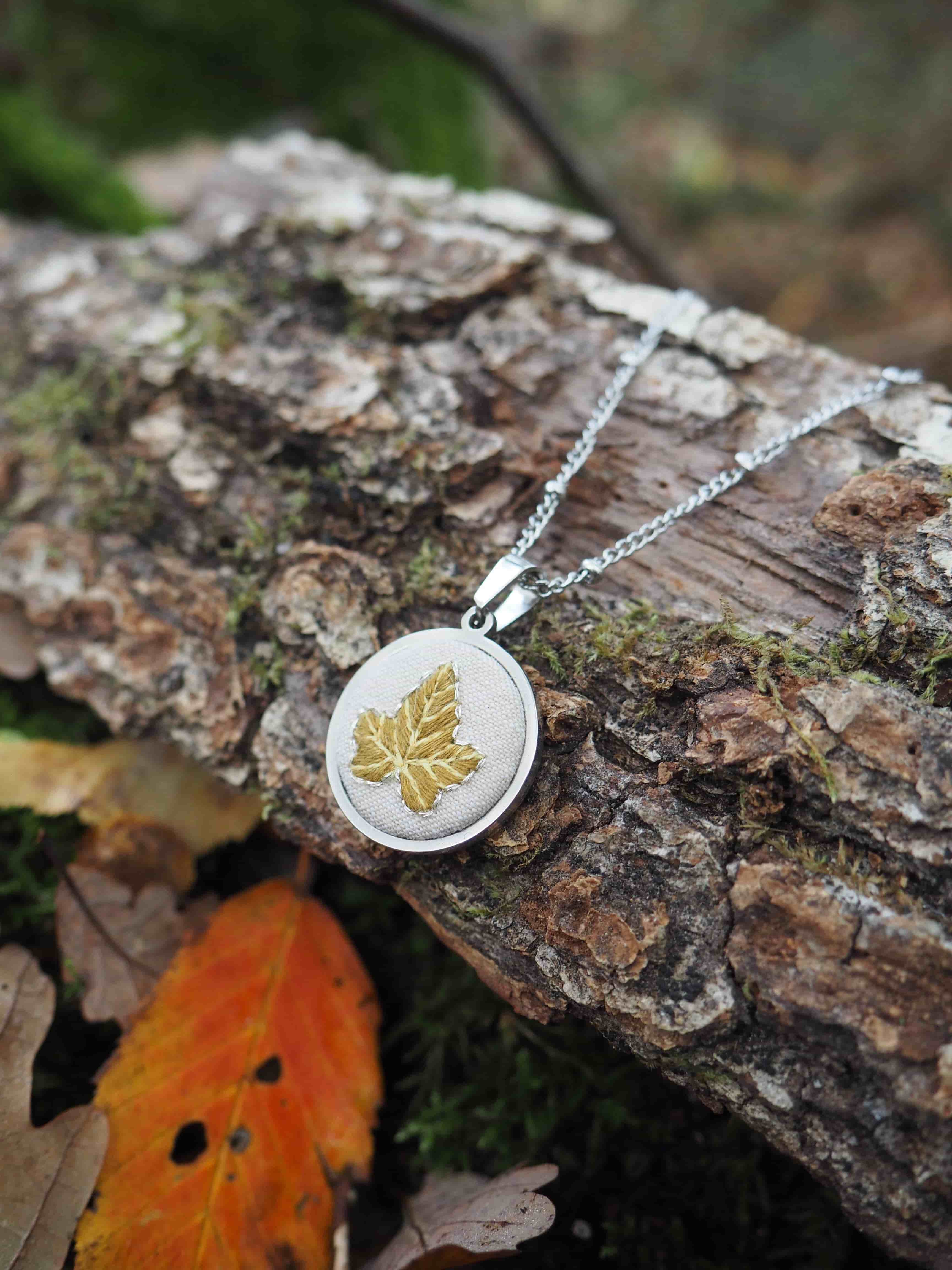 Cette image présente un bijou élégant photographié dans un cadre naturel automnal. Au centre de la composition se trouve un collier avec un pendentif circulaire en métal argenté, orné d'un motif de feuille d'érable doré gravé ou incrusté au centre. Le pendentif est suspendu à une chaîne fine de couleur argentée. Le bijou repose sur un tronc d'arbre ou une bûche dont l'écorce rugueuse et texturée présente des tons bruns naturels avec quelques traces de mousse verdâtre. L'environnement suggère un cadre forestier ou de jardin, avec un arrière-plan flou aux teintes vertes qui évoque la végétation. Une feuille d'automne aux couleurs vives - orange et rouge - est visible dans le coin inférieur de l'image, ajoutant une touche de couleur chaude qui fait écho au motif doré du pendentif. Cette mise en scène crée une harmonie thématique autour du thème automnal et de la nature. L'éclairage naturel met en valeur les détails du bijou tout en créant une atmosphère douce et organique, parfaite pour présenter ce type d'accessoire inspiré de la nature.