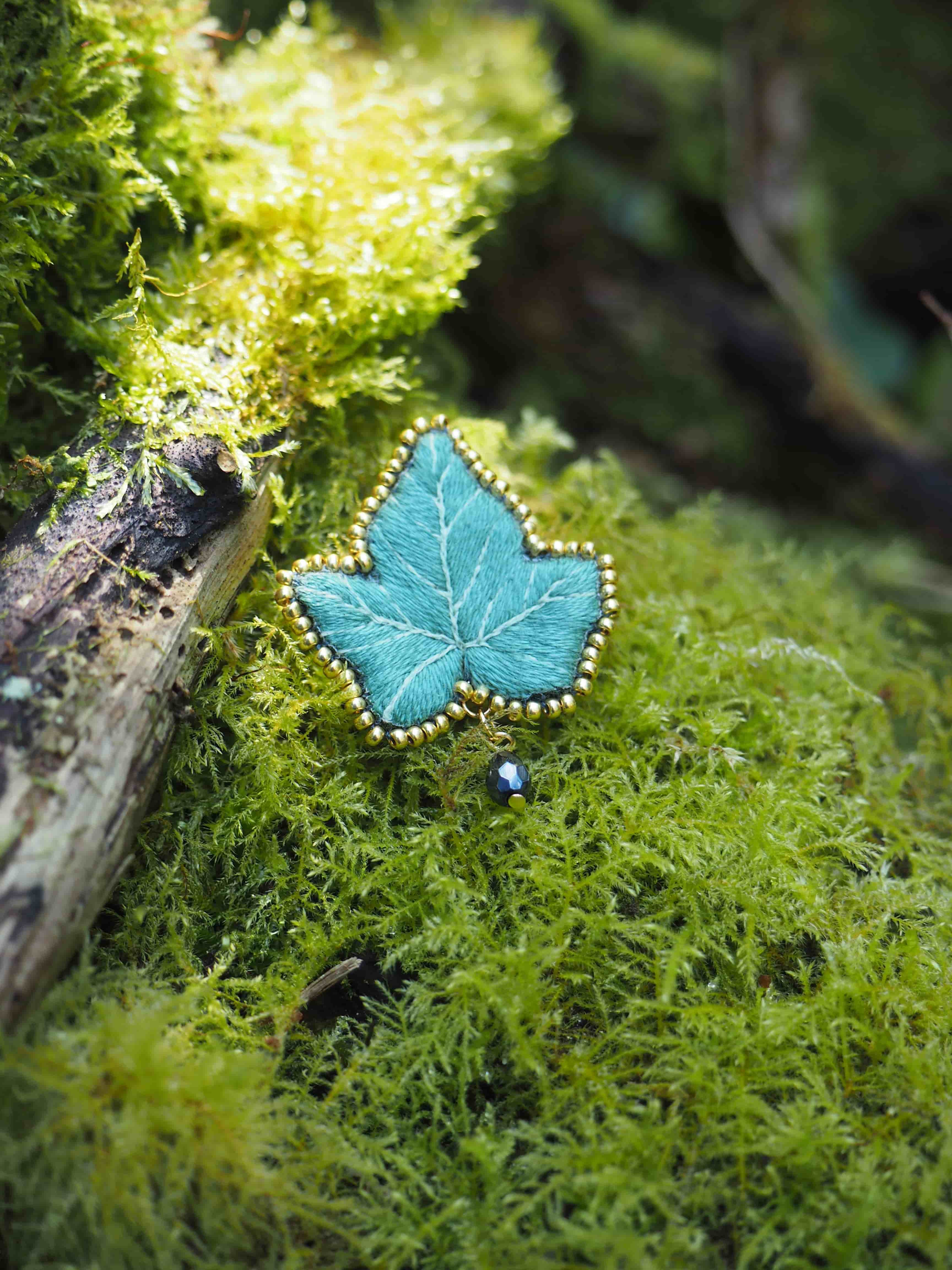 Cette image montre une jolie broche ou un pendentif en forme de feuille, de couleur turquoise, posé sur un lit de mousse verte et luxuriante. La feuille semble être faite d'un matériau émaillé ou peint, et est bordée de petites perles blanches qui lui donnent un aspect délicat et précieux. Le contraste entre le bleu vif de la feuille et le vert tendre de la mousse crée une composition naturelle et poétique. L'arrière-plan flou et la présence d'un petit morceau de bois ajoutent à l'ambiance forestière et féerique de la scène.