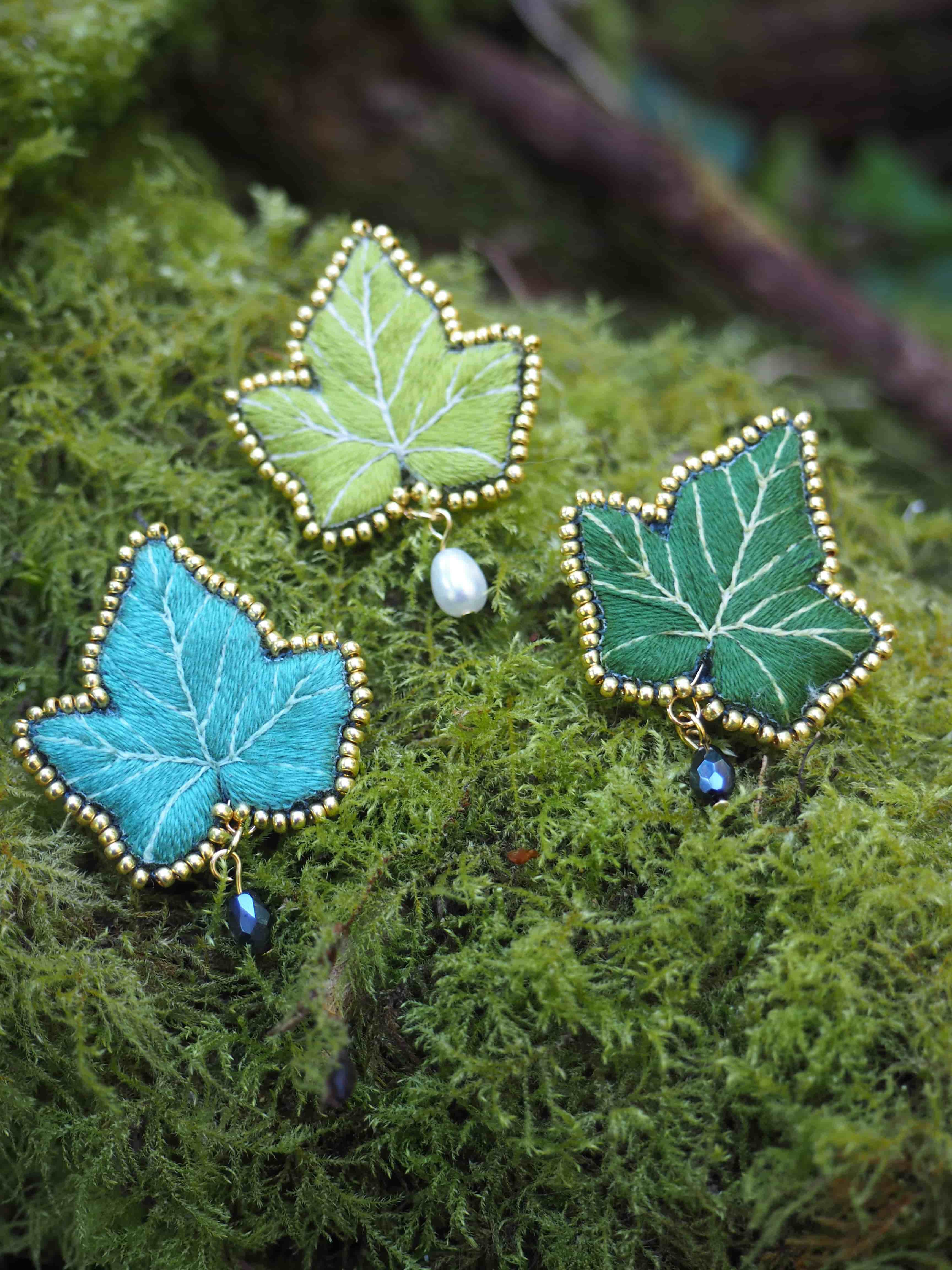 Cette image montre trois jolies broches ou pins artisanaux en forme de feuilles d'érable. Chaque feuille est brodée avec des fils de différentes teintes de vert et bleu : une feuille turquoise, une vert clair et une vert foncé. Les contours des feuilles sont délicatement ornés de petites perles dorées qui créent un effet décoratif élégant. De petites perles (qui semblent être blanches et bleues) sont également ajoutées comme accents. Les broches sont photographiées sur un fond de mousse naturelle verte, ce qui crée une belle mise en scène naturelle et met en valeur ces créations artisanales. Le travail est très détaillé et délicat, montrant une belle maîtrise de la broderie et du travail des perles.