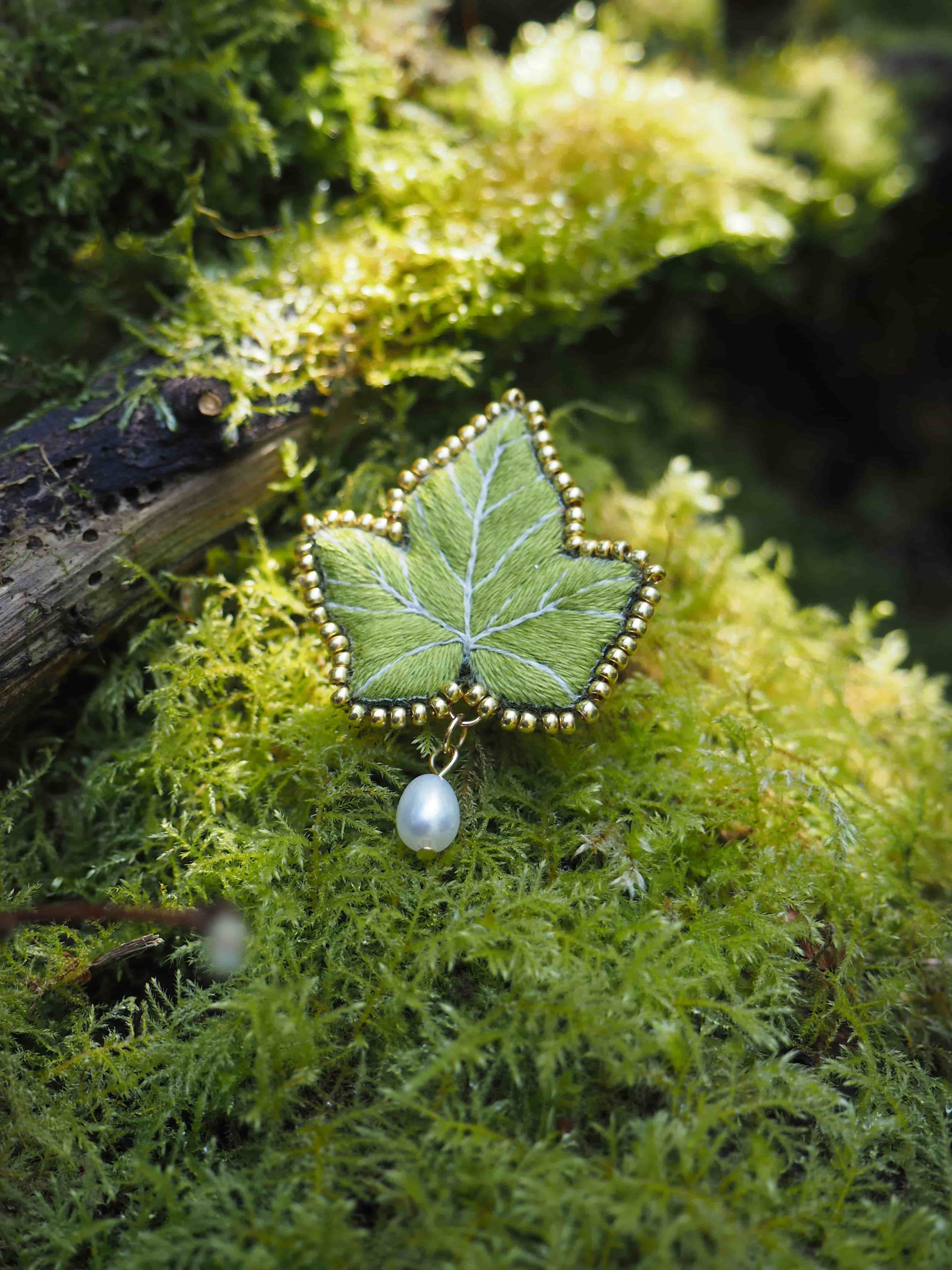Cette image montre un magnifique bijou en forme de feuille posé sur un lit de mousse verte. Le bijou semble être une broche ou un pendentif qui représente une feuille d'érable délicatement ouvragée en vert, bordée de petites perles dorées. Une perle blanche nacrée est suspendue en dessous de la feuille. L'arrière-plan est composé d'une mousse luxuriante d'un vert vif, créant un cadre naturel parfait pour mettre en valeur ce bijou délicat. La photographie capture bien les détails de l'artisanat et crée une ambiance féerique et naturelle avec la combinaison du bijou et de son environnement moussu.