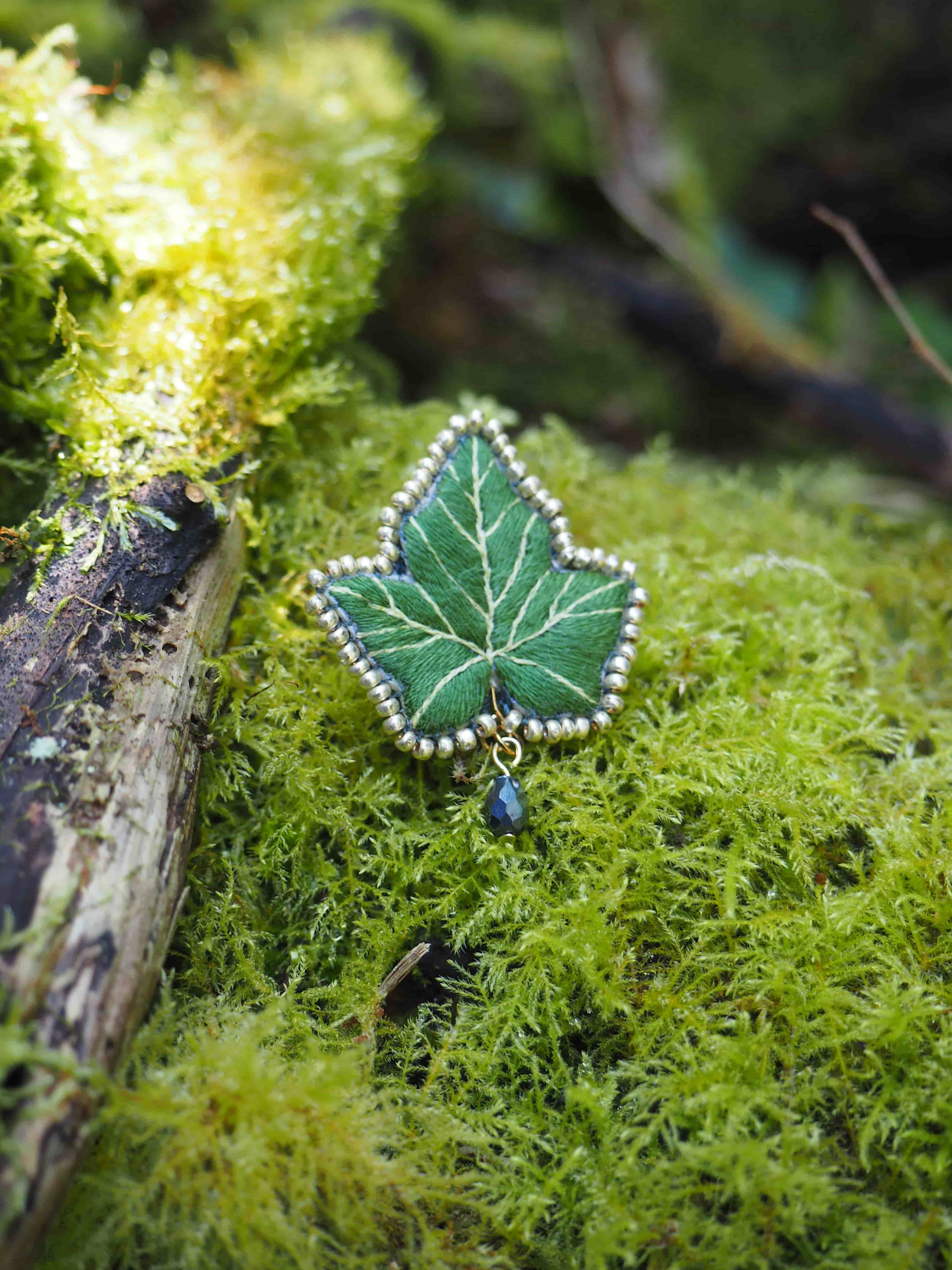 Cette image montre une belle broche ou un pendentif en forme de feuille de lierre, posé sur un lit de mousse verte. Le bijou est d'un vert émeraude et est délicatement bordé de ce qui semble être des petites perles argentées suivant le contour de la feuille. Un petit ornement bleu, probablement une perle ou une pierre, pend au bas de la feuille. La composition est très naturelle et féerique, avec le contraste entre le bijou artisanal et la mousse organique en arrière-plan. On peut également apercevoir un petit morceau de bois ou une brindille sur le côté. L'ensemble crée une atmosphère enchantée, comme si on avait découvert un trésor elfique dans une forêt mystérieuse. La photographie est bien mise au point, mettant en valeur les détails du bijou tout en gardant la texture douce et floue de la mousse en arrière-plan.