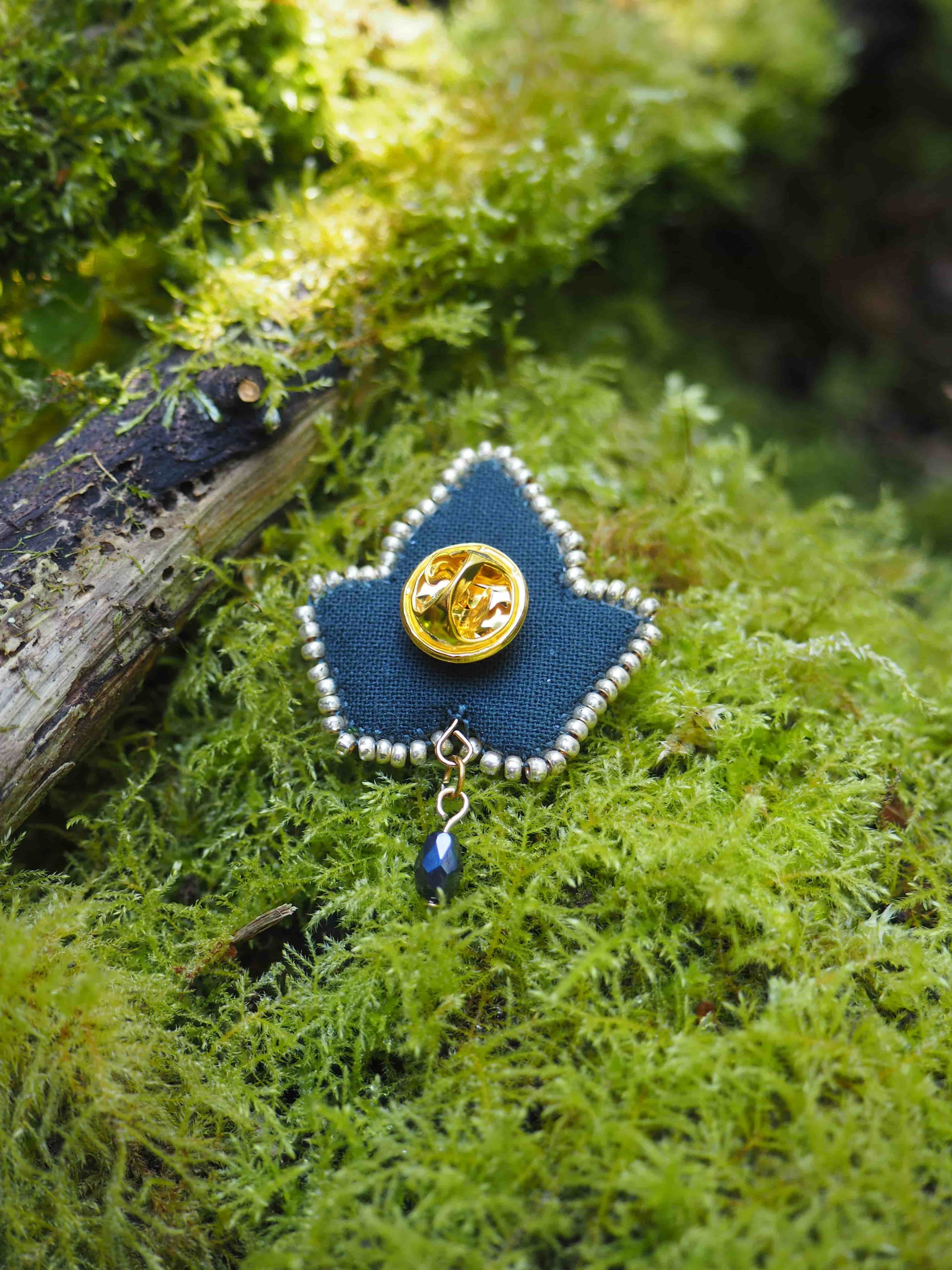 Cette image montre une jolie broche artisanale posée sur un lit de mousse verte. La broche est faite de tissu bleu marine et bordée de petites perles blanches qui forment un contour délicat. Au centre de la broche se trouve un pin's ou un bouton doré, et un petit pendentif bleu est attaché au bas. L'arrière-plan naturel avec la mousse verte luxuriante crée une belle mise en scène qui met en valeur les détails délicats de ce bijou fait main. La composition a un aspect très organique et artistique.