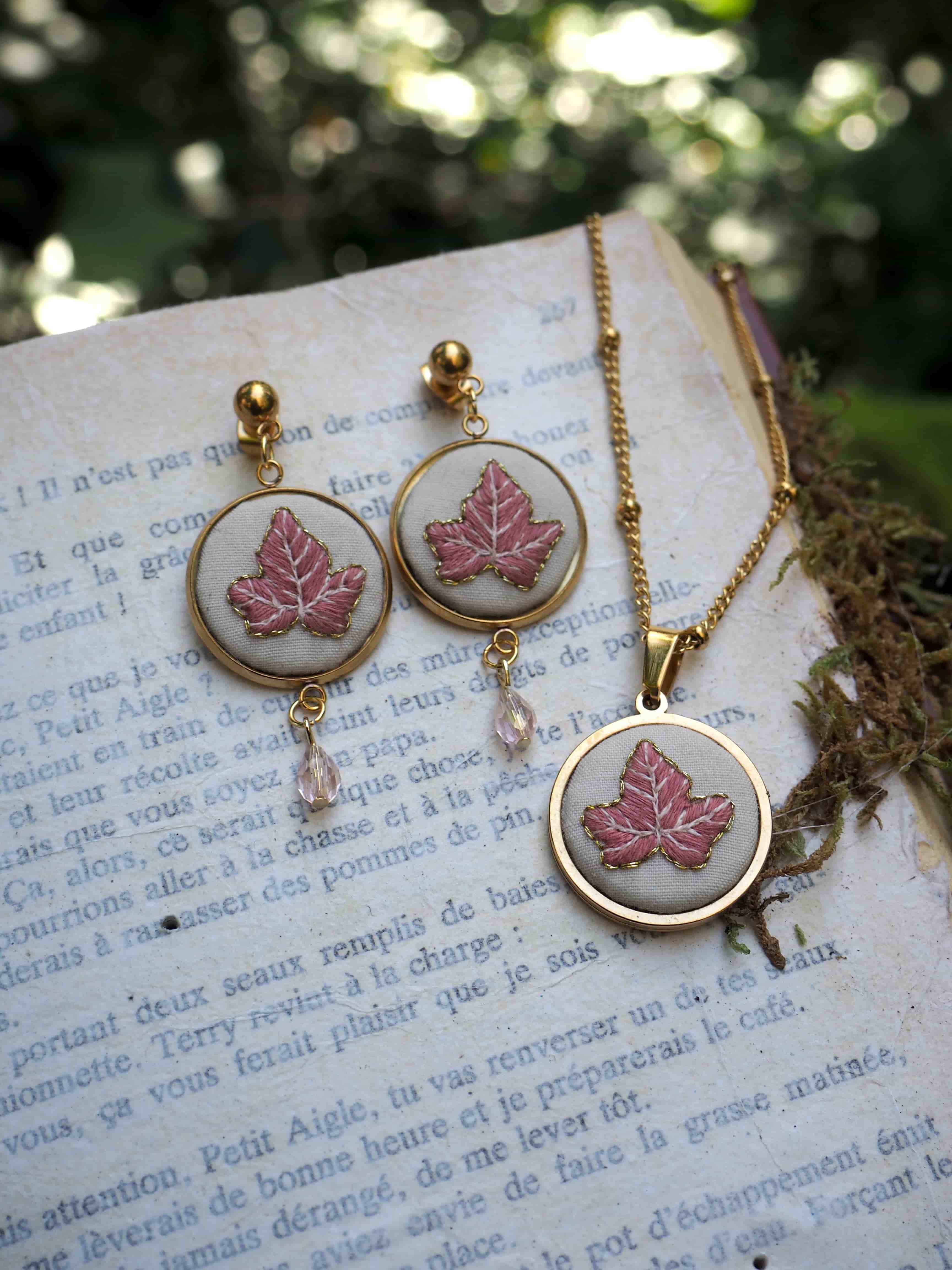 This is a lovely piece of jewelry set featuring matching earrings and a necklace. The design showcases pink maple leaves encased in circular gold-toned frames with a white background. The earrings are drop-style with small crystal or glass beads attached at the bottom, and they have gold ball studs at the top. The pendant on the necklace matches the earrings' design and hangs from a delicate gold chain. The jewelry set is photographed on what appears to be a page of French text, creating a romantic, vintage-inspired aesthetic. The background shows some soft, blurred foliage, adding to the whimsical nature of the composition.