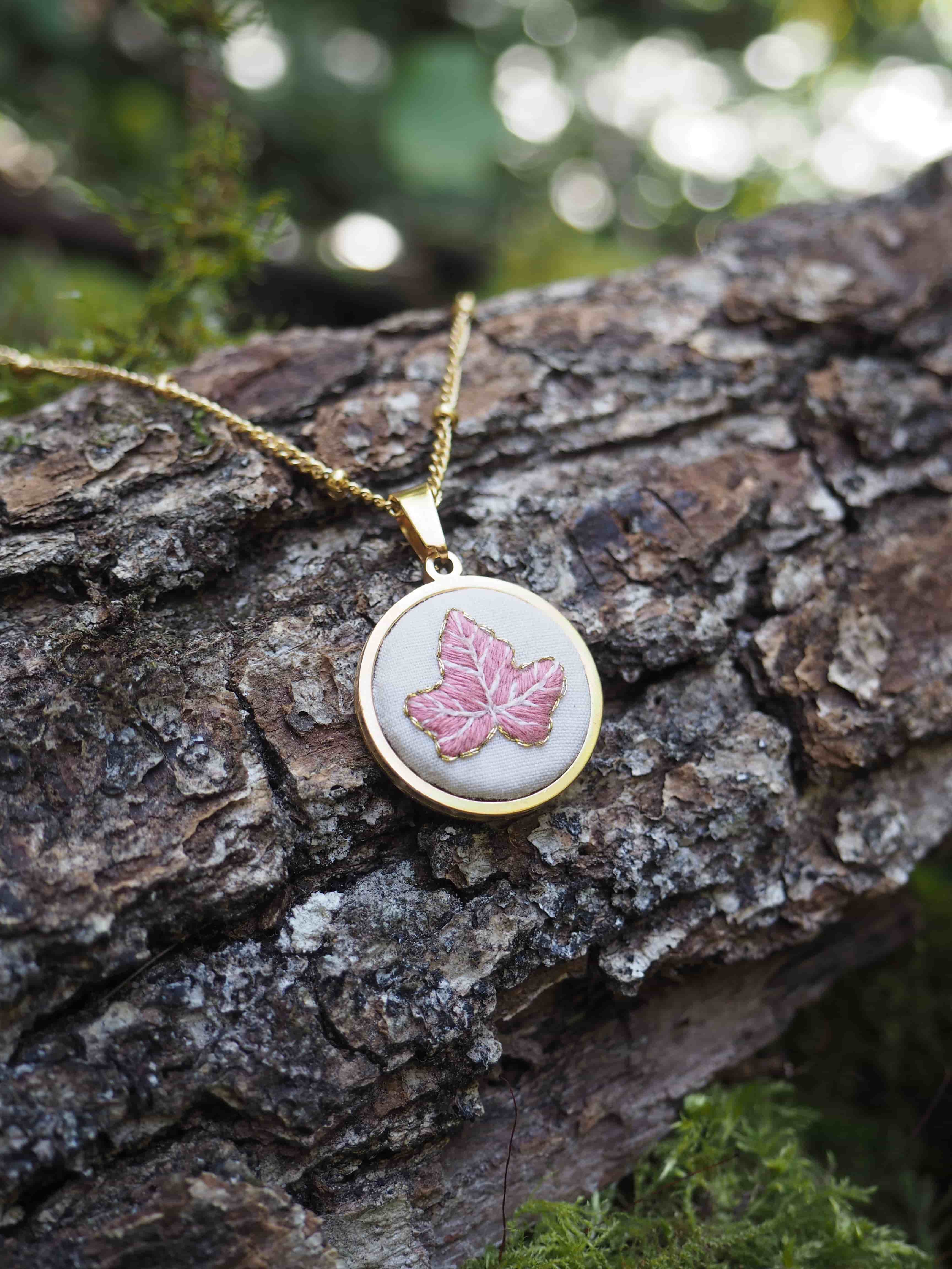 Cette image montre un délicat pendentif posé sur un tronc d'arbre couvert d'écorce rugueuse. Le pendentif est rond, monté sur une chaîne dorée, et présente un motif de feuille d'érable rose sur un fond clair. La composition est artistique avec un bel effet de bokeh (flou) en arrière-plan créant des points de lumière verts et doux. Il y a également un peu de mousse verte visible sur l'écorce, ajoutant à l'ambiance naturelle et forestière de la photo. Le contraste entre le bijou délicat et la texture rugueuse de l'écorce crée une belle composition photographique.