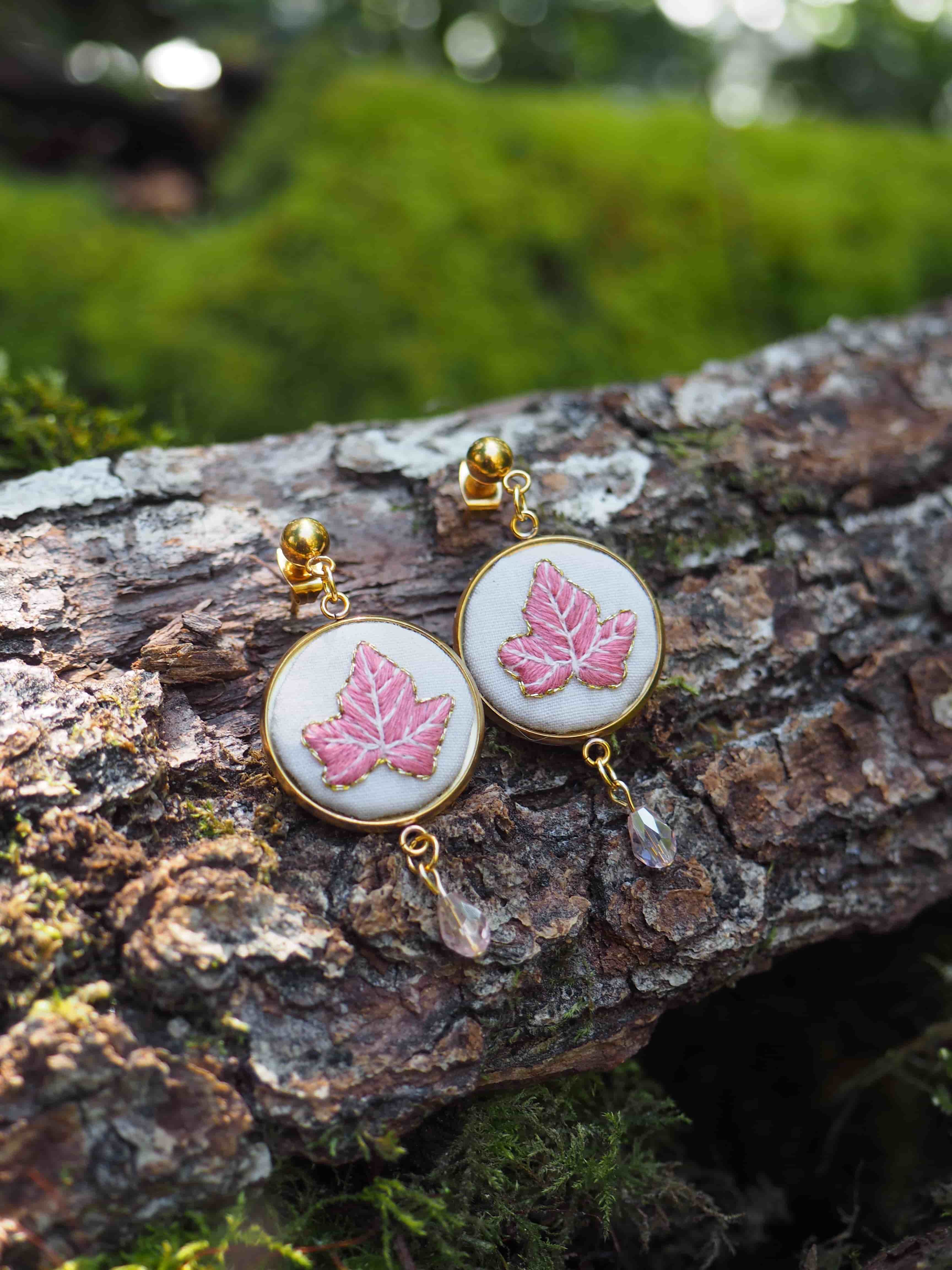 Cette image montre une paire de boucles d'oreilles délicates posées sur un morceau d'écorce d'arbre couvert de mousse. Les boucles d'oreilles sont composées de médaillons ronds avec un cadre doré, et chaque médaillon présente une feuille d'érable rose brodée ou peinte sur un fond blanc. Au bas de chaque boucle d'oreille pend un petit cristal transparent. L'arrière-plan est flou mais on peut voir de la verdure, créant une ambiance naturelle et forestière. La photographie est très artistique, mettant en valeur ces bijoux délicats dans un cadre naturel.