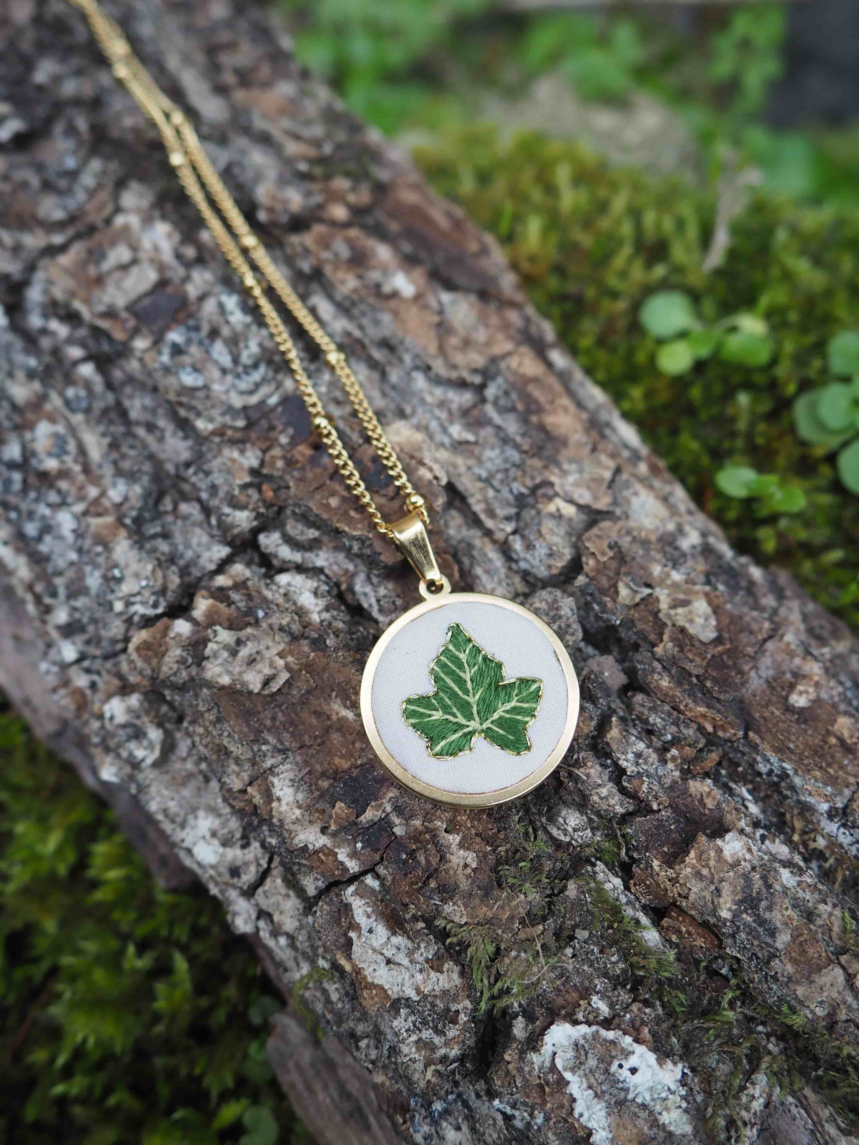 Cette image montre un délicat pendentif sur une chaîne dorée, posé sur un morceau de bois couvert de mousse. Le pendentif est de forme ronde et semble être en émail blanc ou en porcelaine, décoré d'une feuille d'érable verte. La composition est très naturelle et harmonieuse, avec le contraste entre le bijou raffiné et la texture rugueuse du bois et la mousse verte tendre. C'est une belle mise en scène qui évoque la nature et l'artisanat.