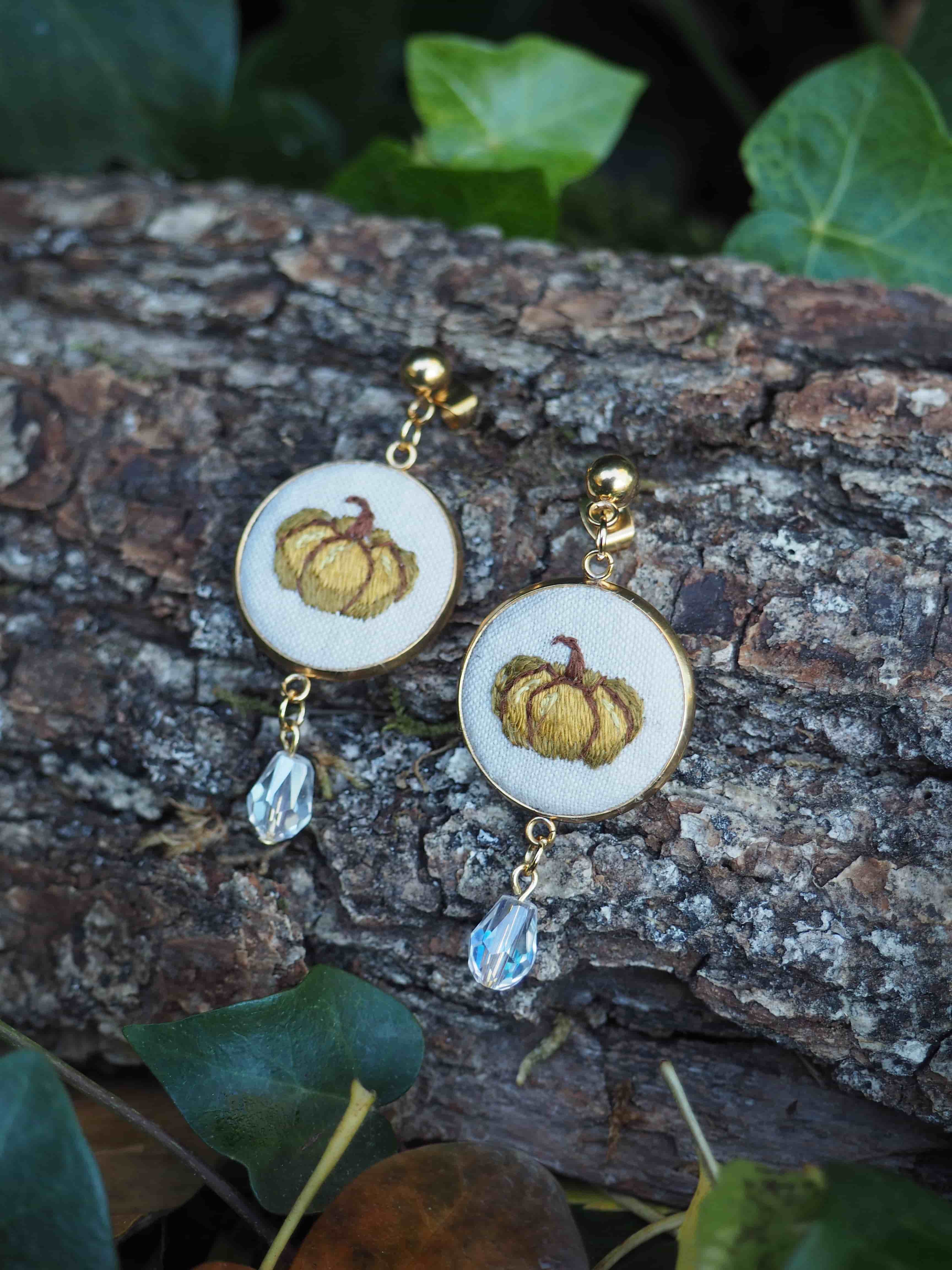 Cette image présente une paire de boucles d'oreilles artisanales disposées sur une surface d'écorce naturelle. Les bijoux se composent d'éléments circulaires blancs ornés de motifs brodés représentant des citrouilles dorées avec des tiges brunes, évoquant un thème automnal. Chaque boucle d'oreille est complétée par une perle cristalline transparente qui pend élégamment en dessous. Les boucles d'oreilles sont dotées de crochets dorés et reposent sur une écorce rugueuse aux tons bruns et gris, créant un contraste naturel avec les bijoux. L'arrière-plan se compose de feuillage vert flou, avec des feuilles aux formes variées qui encadrent délicatement la composition. L'éclairage naturel met en valeur les détails de la broderie et fait scintiller subtilement les perles cristallines. Cette mise en scène champêtre souligne le caractère artisanal et saisonnier de ces accessoires, parfaits pour célébrer l'automne avec élégance.