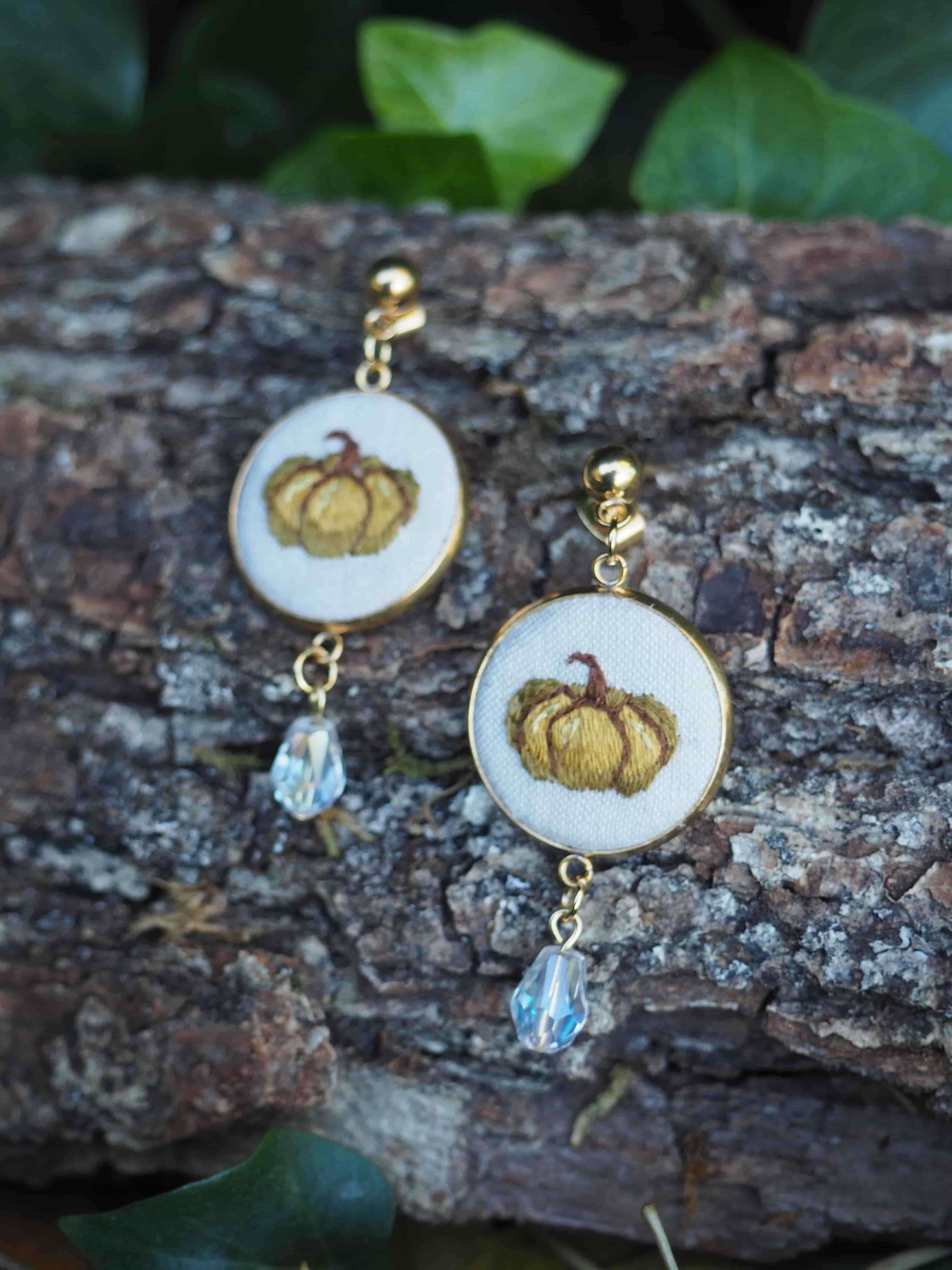 Cette image présente une paire de boucles d'oreilles élégantes disposées sur une surface naturelle en bois brut.  **Caractéristiques des bijoux :**
- **Design principal** : Chaque boucle d'oreille comporte un médaillon ovale blanc avec un motif décoratif doré représentant ce qui semble être une citrouille ou un fruit stylisé
- **Finitions** : Les éléments métalliques sont dorés, donnant un aspect luxueux
- **Pendentifs** : Chaque boucle se termine par une petite perle ou cristal transparent qui accroche la lumière
- **Style** : L'ensemble évoque un style vintage ou artisanal avec une touche automnale **Mise en scène :**
- Les boucles d'oreilles reposent sur une écorce d'arbre texturée, créant un contraste naturel
- L'arrière-plan flou laisse apercevoir quelques feuilles vertes
- L'éclairage naturel met en valeur les détails dorés et la transparence des pendentifs Cette composition photographique met parfaitement en valeur ces bijoux artisanaux dans un cadre naturel et organique.