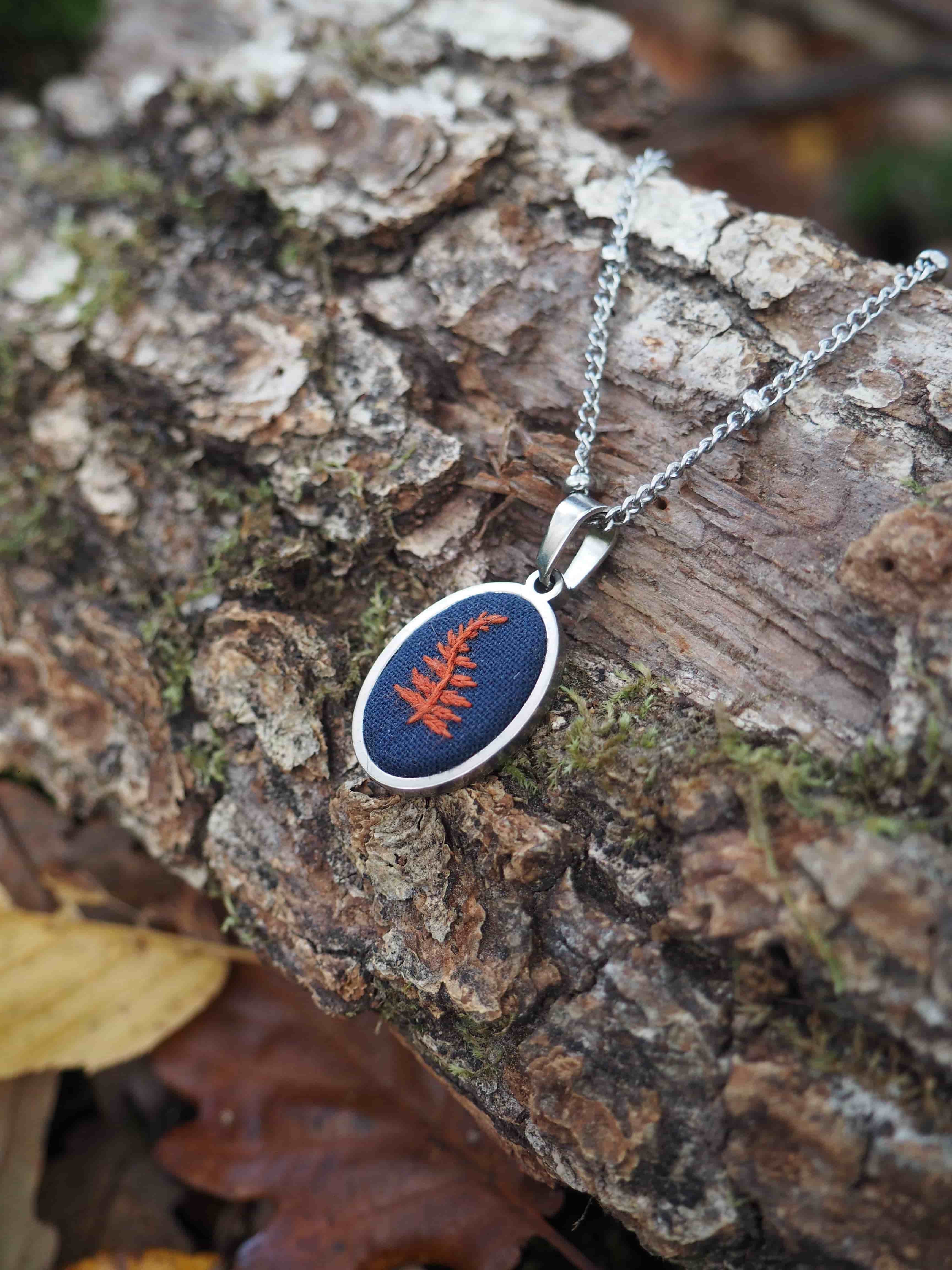 Cette image présente un pendentif élégant posé sur un morceau d'écorce naturelle dans un environnement forestier. Le bijou se compose d'une chaîne argentée délicate reliée à un pendentif de forme ovale avec un cadre métallique argenté. Le pendentif contient une résine ou un matériau transparent de couleur bleue profonde dans lequel est enchâssée une feuille naturelle de couleur rouge-orangé, probablement une fougère ou une petite feuille d'automne. Cette inclusion végétale crée un contraste saisissant avec le fond bleu. L'ensemble repose sur une surface d'écorce rugueuse et texturée, avec des tons bruns et gris naturels. On peut apercevoir quelques mousses verdâtres sur l'écorce, ainsi qu'une feuille jaunâtre en arrière-plan, ce qui renforce l'ambiance naturelle et organique de la composition. L'éclairage met bien en valeur les détails du bijou et crée une atmosphère chaleureuse qui souligne le thème nature de cette pièce artisanale. Ce type de pendentif semble être un bijou artisanal qui capture et préserve un élément naturel dans une résine colorée.