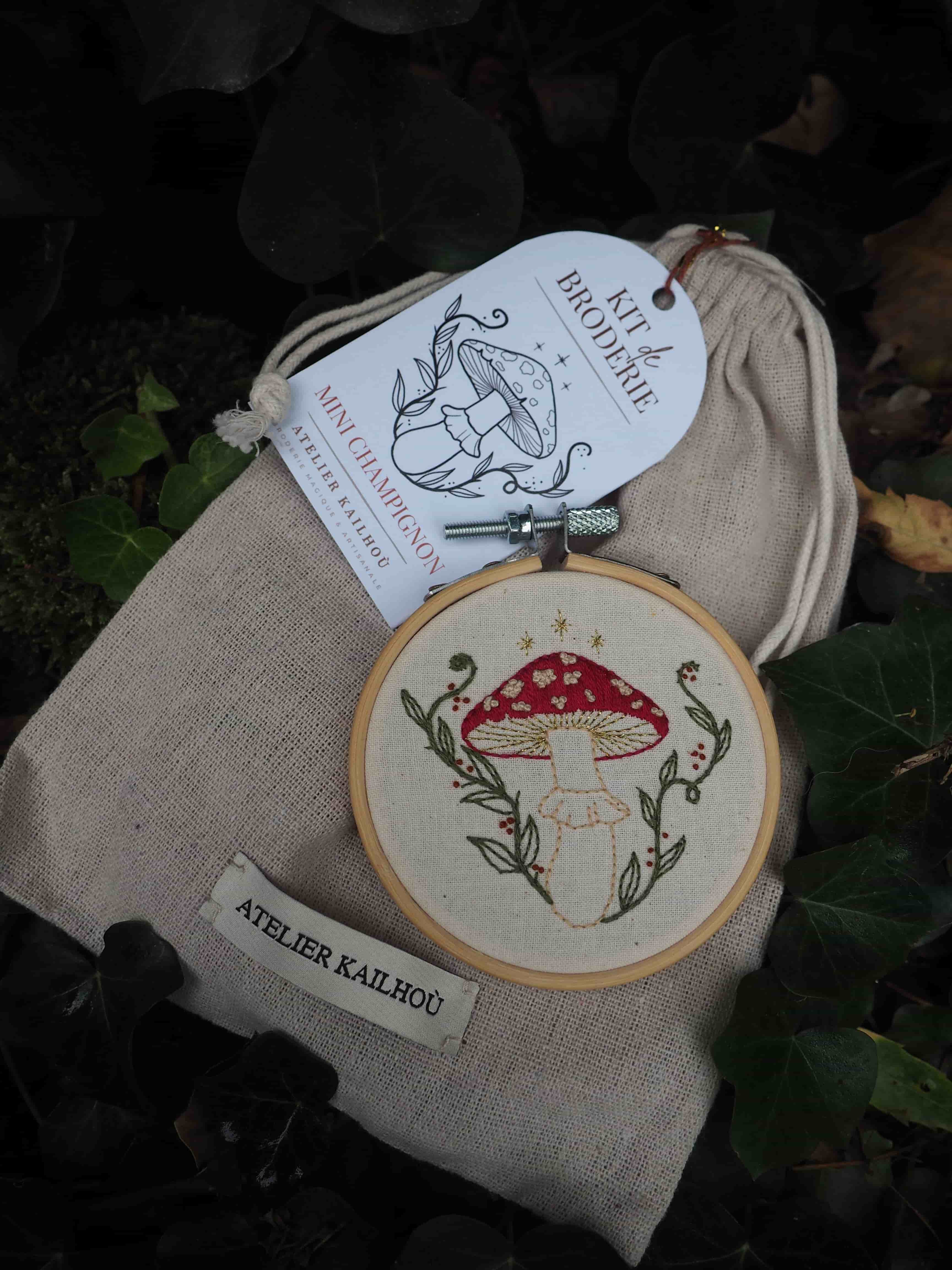 Cette image présente un kit de broderie artisanal disposé sur un fond de feuillage naturel. On peut voir : **L'élément principal** : Un tambour à broder en bois contenant une toile beige sur laquelle est brodé un champignon rouge à pois blancs (amanite tue-mouches) entouré de petites feuilles et brindilles vertes délicatement travaillées. **L'emballage** : Un sac en toile de lin naturelle de couleur beige/écru avec une étiquette blanche portant une illustration similaire au motif brodé et le texte "Kit broderie" ainsi que la mention "ATELIER KAILHOO" en bas. **Le style** : La broderie présente un travail soigné avec des points fins, dans un style naturaliste et délicat. Les couleurs utilisées sont douces - rouge pour le chapeau du champignon, blanc pour les pois, vert tendre pour la végétation. **L'ambiance** : L'ensemble évoque l'artisanat fait main, le travail traditionnel de broderie, avec une esthétique champêtre et naturelle renforcée par le fond de vraies feuilles vertes sur lequel est photographié le kit.