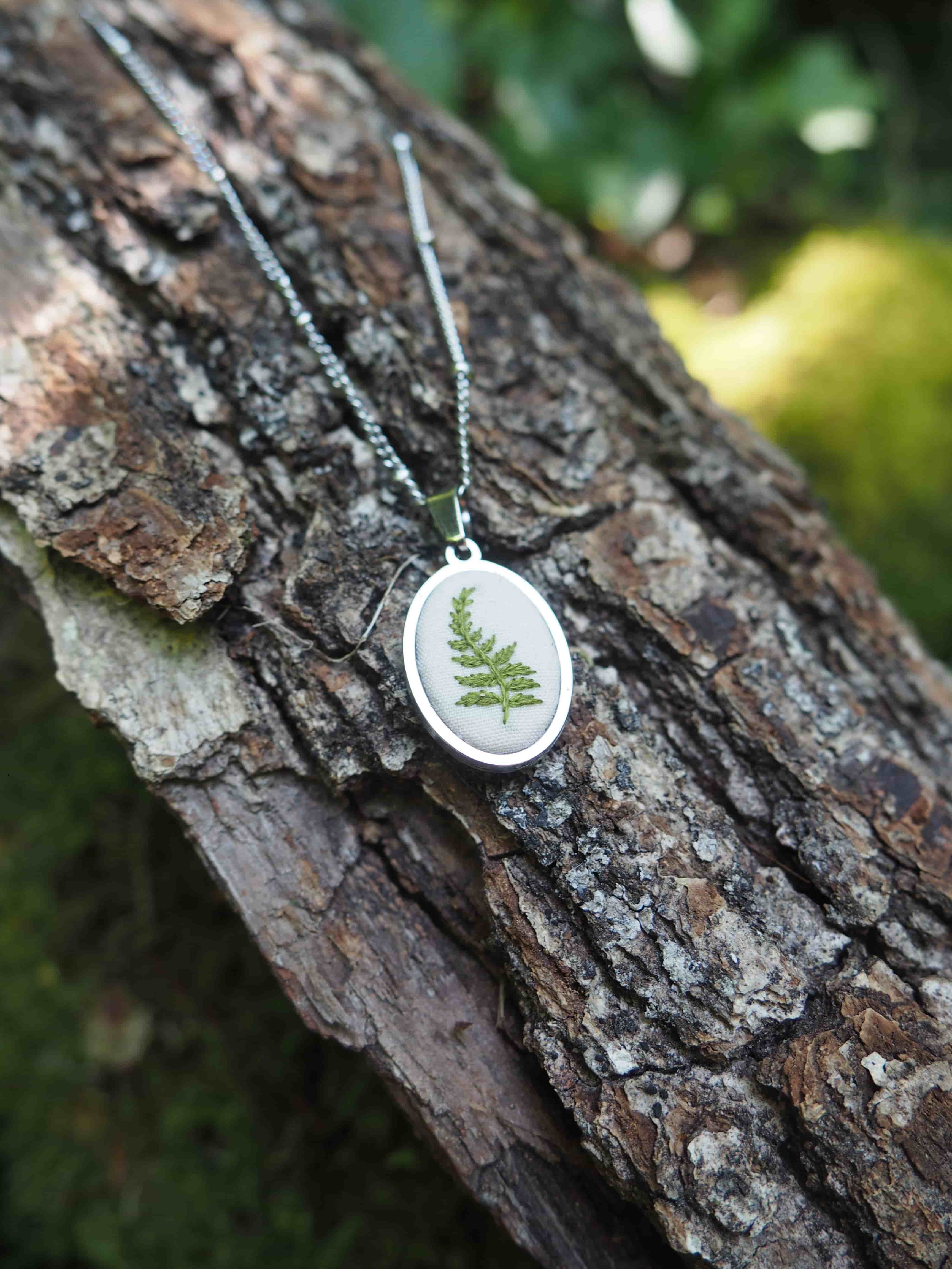 L'image montre des bijoux ou des pendentifs délicats qui semblent contenir de véritables petites fougères préservées. Ces pendentifs sont de forme ronde avec ce qui semble être un cadre blanc ou argenté, et à l'intérieur se trouve une fougère verte naturelle, probablement conservée sous verre ou dans de la résine. Ils sont photographiés sur ce qui semble être l'écorce rugueuse d'un arbre, créant un contraste intéressant entre l'aspect naturel brut de l'écorce et la délicatesse des pendentifs. L'image a une belle composition naturelle et organique qui met en valeur ces bijoux botaniques.