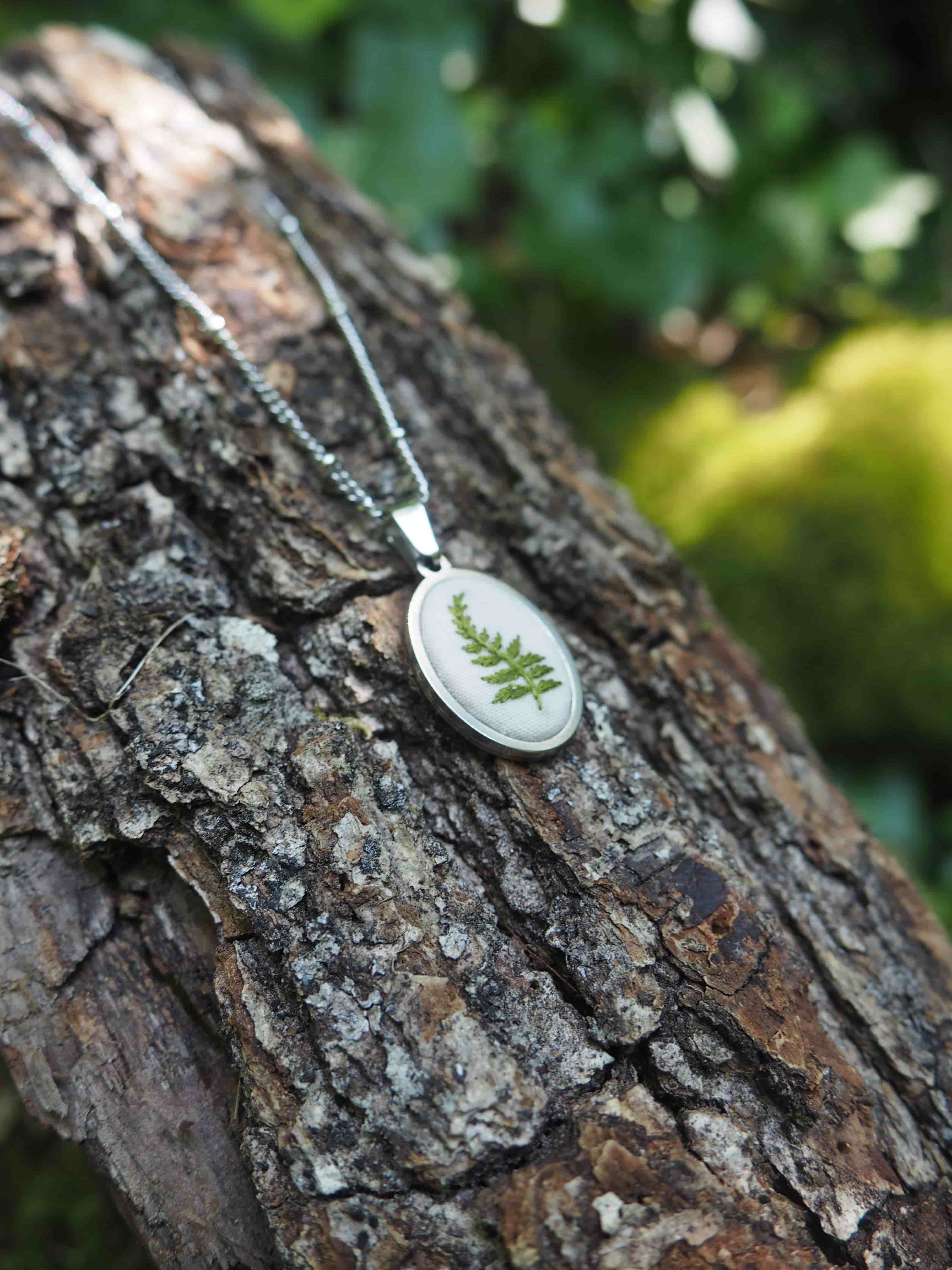 L'image montre un pendentif délicat posé sur l'écorce d'un arbre. Le pendentif semble être en argent et contient une vraie fougère préservée sous verre ou résine dans un médaillon ovale. Il est attaché à une fine chaîne argentée. L'arrière-plan montre l'écorce texturée et rugueuse de l'arbre avec de la mousse verte floue et du feuillage en arrière-plan, créant une belle composition naturelle. C'est un bijou qui capture élégamment un petit morceau de nature.
