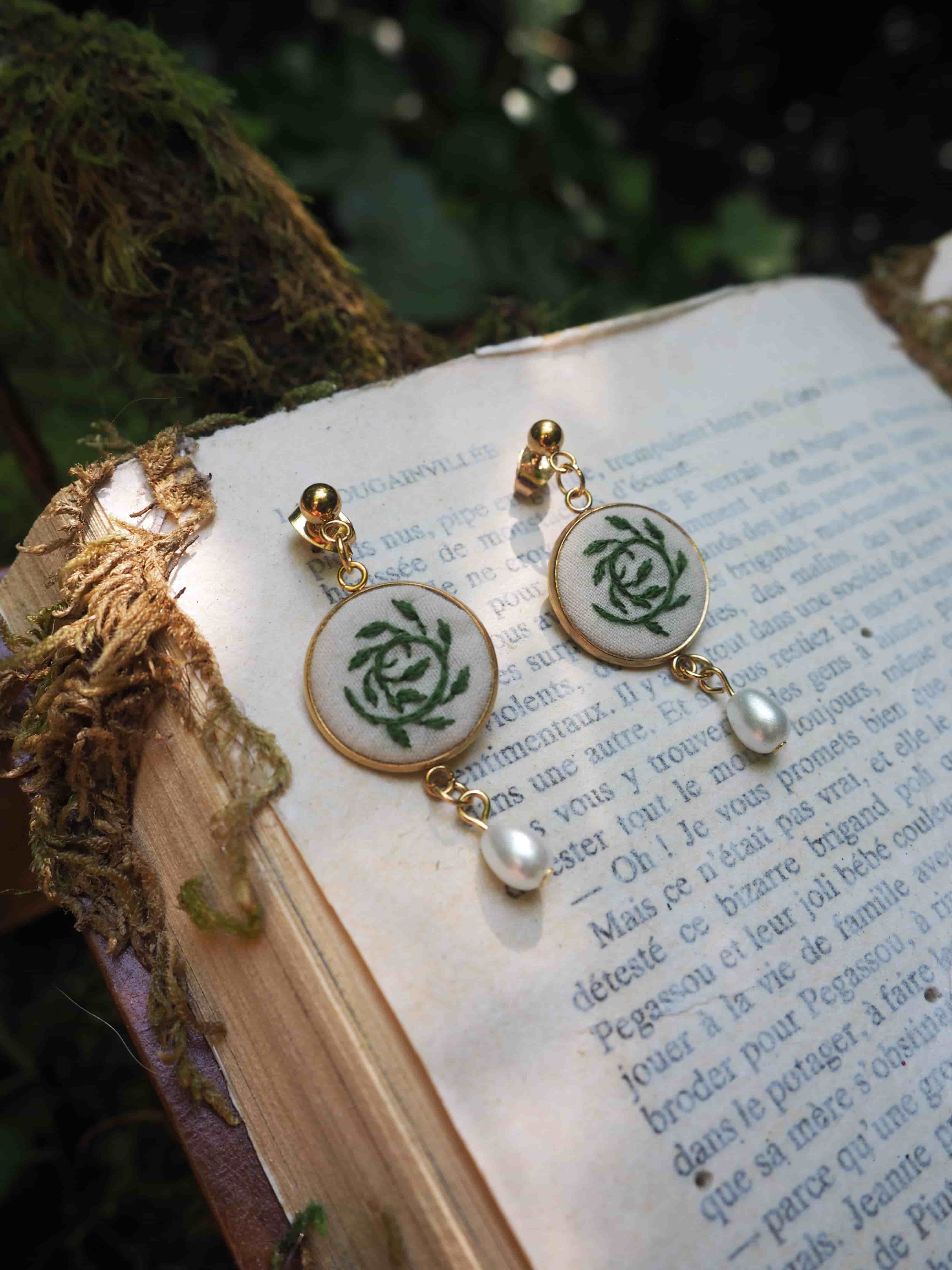 Cette image montre une paire de boucles d'oreilles élégantes posées sur un livre ouvert. Les boucles d'oreilles sont composées de médaillons ronds avec un motif de rose stylisée en vert sur fond blanc, encadrés d'une monture dorée. Chaque boucle d'oreille est ornée d'une petite perle blanche qui pend en dessous du médaillon. Le livre sur lequel elles reposent semble être un texte en français, et on peut apercevoir de la mousse ou de la végétation verte floue en arrière-plan, donnant à l'image une ambiance romantique et vintage. L'ensemble crée une composition délicate et poétique.